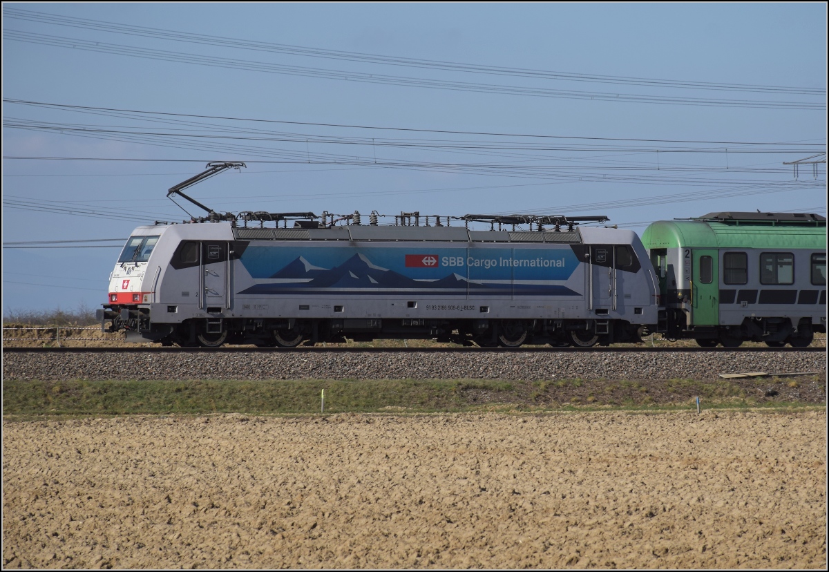 Auch eine Kombination: Eine Lok der BLS Cargo mit Werbung des Konkurrenten SBB, immatrikuliert und ein Produkt aus Kassel... 
91 83 2186 908-6 I-BLSC lautet die Immatrikulationsnummer der Lok, hier zu sehen zwischen den Feldern bei Auggen. Saharastaub hat den Blick ins Elsass getrübt. März 2021