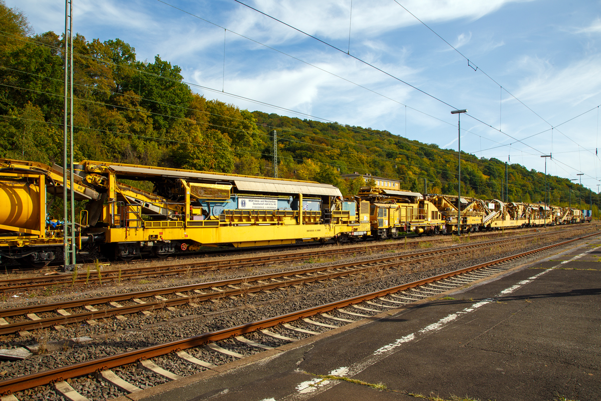 
Bild 1
Die Plasser & Theurer Bettungsreinigungsmaschine RM 95-800 W der MGW Gleis- und Weichenbau-Gesellschaft mbH & Co. KG (Berlin) ist am 20.09.2020 im Bahnhof Dillenburg abgestellt.

Hier ein Überblick über die eigentliche Maschine. Von links nach rechts die Abwasserkläranlage 99 80 9510 002-5 D-MGWB und die eigentlichen Bettungsreinigungsmaschine RM 95-800 W (99 80 9414 001-4 D-MWGB), sowie am Zugende noch der Schutzwagen 3999 010-6 der Gattung Res.

Die Grundmaschine wurde 2010 von Plasser & Theurer unter den Fabriknummern 5221 bis 5224 gebaut, sie hat die VDM Nr. 99 80 9414 001-4 D-MWGB.

Bei der RM 95-800 W von Plasser & Theurer handelt es sich um eine Hochleistungsbettungsreinigungsmaschine mit integriertem Schotterwaschmodul und einer Abwasserkläranlage.

Im Rahmen der kompletten Schotteraufbereitung wird das sogenannte Feinkorn separiert und nur noch dieses muss abtransportiert werden. Der zur Wiederverwendung vorgesehene Schotter wird gewaschen, wieder angeschärft und anschließend in das Gleisbett eingebracht. Durch diese Maßnahmen werden eine bessere Qualität des Schotterbetts und eine längere Liegedauer gewährleistet. Optional kann jedoch auch Neuschotter zugemischt werden. Durch diese besondere Konzeption der Maschine tritt nicht nur eine erhebliche Kosteneinsparung ein, sondern auch die mit dem geringeren Transportaufwand verbundenen Emissionen werden erheblich reduziert. 

TECHNISCHE DATEN der RM 95-800 W: 
Gesamtgewicht: 427.000 kg 
Länge über Puffer: 114.980 mm (ohne Klärwagen) 
Drehzapfenabstände: 15.000/ 25.500/ 14.500/ 16.000/10.000/10.500 mm 
Höhe: 4.295 mm
Breite: 3.250 mm
Achsanzahl: 22
Höchstgeschwindigkeit : 19 km/h (Eigenfahrt) / 100 km/h (geschleppt):
Reinigungsleistung: bis 300 m³/h bei Reinigung/Vollaushub und Waschbetrieb.