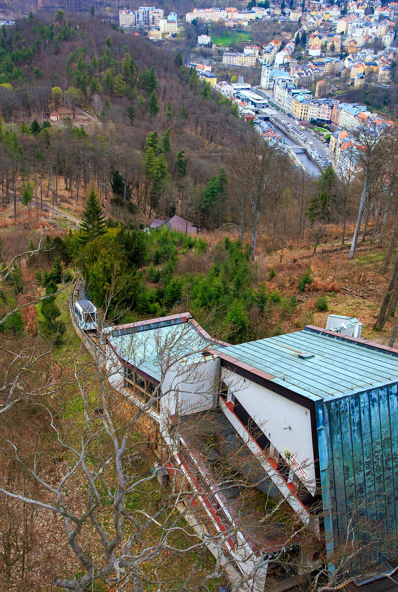 Blick auf die Bergstation der Standseilbahn Diana (tschechisch Lanová dráha Diana) und im Tal aus Karlsbad (Karlovy Vary), vom 40 m hohen Diana-Aussichtsturm auf der Freundschaftshöhe 556 m ü.M. am 18.04.2023. Der Wagen „DIANA 1“ der Standseilbahn fährt gerade wieder hinab.

Die Stadtseilbahnen (Diana sowie Imperial) stellen in Karlsbad schon seit Anfang des vergangenen Jahrhunderts ein traditionelles und beliebtes Verkehrsmittel dar. Sie gehören inzwischen zum Stadtbild und sind ein beliebtes Touristenziel.

Diana-Standseilbahn:
Die Standseilbahn Diana (tschechisch Lanová dráha Diana) wurde im August 1912 eröffnet und beginnt an der Station Alte Wiese (Stará louka) auf einer Höhe von 389 m und führt auf die Freundschaftshöhe 556 m über NN, wo sich der Diana-Aussichtsturm befindet. Die Talstation befindet sich gleich neben dem Grandhotel Pupp in der Gasse Mariánská. Den Höhenunterschied von 167 m überwindet sie auf einer Strecke von 437 m. Im Bereich der abtsche Ausweiche liegt auch die Mittelstation Jelení skok (Hirschsprung) in 473 m Höhe. Die maximale Steigung beträgt 432,3 ‰, durchschnittlich liegt diese bei 398,1 ‰. Die Bahn wurde insgesamt dreimal renoviert, zwischen 1963 und 1965, 1972 und noch einmal zwischen 1984 und 1988. Im Gegensatz zur Bahn zum Personennahverkehrs der Stadt. Auch sie wird im 15-Minuten-Takt befahren und benötigt 4 Minuten für eine Fahrt bei einer Geschwindigkeit von 2,04 m/s. Die Gleise sind vom Typ A, die Spurweite beträgt 1.000 mm. Die leer 7.900 kg wiegenden Wagen sind für 49 + 1 Fahrer ausgelegt. Das Seil hat einen Durchmesser von 28 mm. Die Antriebsleistung beträgt 51 kW, die Antriebsregulation erfolgt mit Hilfe von Thyristoren, 

Die Seilbahn ist ein Projekt des Ing. H. H. Peter aus Zürich. Der Bau der Seilbahn wurde von der Wiener Baufirma Leo Arnoldi mit Hilfe von lokalen Handwerkern und Firmen durchgeführt. Die erste Generalüberholung der Seilbahn geschah in den Jahren 1963-1965, als die Seilbahnen neue Metallwagons aus der Produktion des Tatra Smíchov Wagon bekamen. Die Rekonstruktion der elektrischen Ausrüstung wurde 1972-73 durchgeführt.Es ist ein traditionelles und sehr beliebtes Verkehrsmittel auf der Freundschaftshöhe in Karlsbad, das hauptsächlich von den Touristen der Stadt genutzt wird. Von der Talstation wird man zum Gipfel gebracht, wo sich der Aussichtsturm Diana und das Wald-Restaurant und Café Diana befindet.

Betreiber der Seilbahnen ist die DPKV - Dopravní podnik Karlovy Vary a. s. (Verkehrsbetrieb Karlsbad AG). Der Verkehrsbetrieb Karlsbad AG ist ein traditioneller Betreiber des öffentlichen Verkehrs in Karlsbad. Er betreibt tagsüber und auch in den Nachtstunden die regelmäßigen Stadtbusverkehrslinien, Sonderverkehrslinien und Vorortverkehrslinien. Ein Besonderheit des Karlsbader ÖÖV ist der Verkehr der Stadtseilbahn vom Theaterplatz (Divadelní náměstí) zum Hotel Imperial und vom Grandhotel Pupp zum Aussichtsturm Diana. Der Verkehrsbetrieb Karlsbad AG bietet der Öffentlichkeit auch weitere, überwiegend mit der Beförderung zusammenhängende Dienstleistungen an. 

DATEN der Seilbahn DIANA:
Höhenunterschied: 167 m
Streckenlänge:  453 m
Spurweite: 1000 mm (Meterspur)
Maximale Steigung:  432,3 ‰
Leergewicht eines Wagens: 7.900 kg
Kapazität eines Wagens: 49 + 1 Fahrer
Seildurchmesser: 28 mm
Antriebsleistung: 51 kW
Höchstgeschwindigkeit: 7,3 km/h
Taktung: alle 15 Minuten
Fahrtdauer: ca. 3 Minuten
Preis (z.Z.) für Hin- und Rückfahrt: 150 CZK