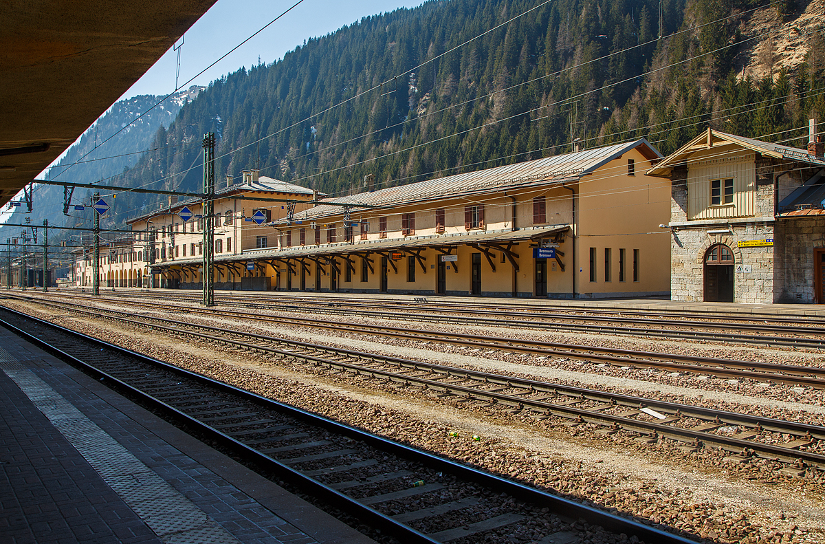 Der Bahnhof Brenner (Stazione di Brennero) am 27.03.2022, der Blick vom Bahnsteig 6 in s�dlicher Richtung aufs Empfangsgeb�ude. 

Der Bahnhof Brenner (italienisch Stazione di Brennero) befindet sich an der Brennerbahn. Er ist der Grenzbahnhof zwischen Nord- und S�dtirol und so zwischen �sterreich und Italien. Der Bahnhof liegt auf der Passh�he des Brenners (1.370 m). Dieser enge, h�chstgelegene Abschnitt des Wipptals bietet zwischen steilen Bergh�ngen nur wenig Platz f�r das kleine Passdorf und wird zu gro�en Teilen von Verkehrsinfrastrukturen wie dem Bahnhof, der A22/A 13 und der SS 12/B 182 beansprucht. Der Bahnhof befindet sich fast zur G�nze auf dem Gebiet der S�dtiroler Gemeinde Brenner bzw. auf italienischem Staatsgebiet, das hier auch auf Fl�chen n�rdlich der Wasserscheide �bergreift. Lediglich kleinere Teile im n�rdlichen Bereich des Bahnhofs geh�ren zur Tiroler Gemeinde Gries bzw. zum �sterreichischen Staatsgebiet.

Der von Wilhelm von Flattich entworfene Bahnhof Brenner wurde 1867 zusammen mit dem gesamten Abschnitt der Brennerbahn zwischen Innsbruck und Bozen in Betrieb genommen, damals geh�rte ja noch alles zur Doppelmonarchie �sterreich-Ungarn. An den planenden Ingenieur des Projekts, Carl von Etzel, erinnert eine Bronzeb�ste am Bahnhofsgel�nde. Die urspr�nglich relativ unbedeutende Station erfuhr 1888 aufgrund der hohen Fahrgastzahlen der neuen Strecke eine erste Erweiterung.

Die Annexion S�dtirols durch Italien infolge des Ersten Weltkriegs ver�nderte die Situation grundlegend, da nun die neue Staatsgrenze �ber den Brenner verlief. Die Teilstrecken Innsbruck–Brenner und Verona–Brenner wurden von nun an von zwei verschiedenen Verwaltungen betreut, die sich zudem noch in feindseliger Haltung gegen�berstanden. 1930 wurde ein kompletter Neubau des Empfangsgeb�udes eingeweiht.

In der Bildmitte an den OL-Auslegern sieht man die Fahrleitungssignale, 
auf der Spitze stehenden Quadrat mit blauem Hintergrund und wei�em Rand in wei� angebracht, hier der waagerechte wei�e Streifen zeigt so das „Signal - El 4 - B�gel ab (Stromabnehmer tief)“. Das Signal kennzeichnet die Stelle, an der der Oberleitungsb�gel einer Ellok vollst�ndig abgesenkt sein muss. Damit der Lokf�hrer mit dem Absenken rechtzeitig beginnt, ist 250m vor diesem Signal das Signal El3 aufgestellt. 

Hier endet das in �sterreich (�BB) �bliche Stromsystem mit 15 kV, 16,7 Hz Wechselstrom, etwas s�dlicher beginnt dann das in Italien (RFI) �bliche Stromsystem mit 3 kV Gleichstrom. Es zeigt aber hier nicht die Landesgrenze, die liegt weiter n�rdlicher (in meinem R�cken). 
