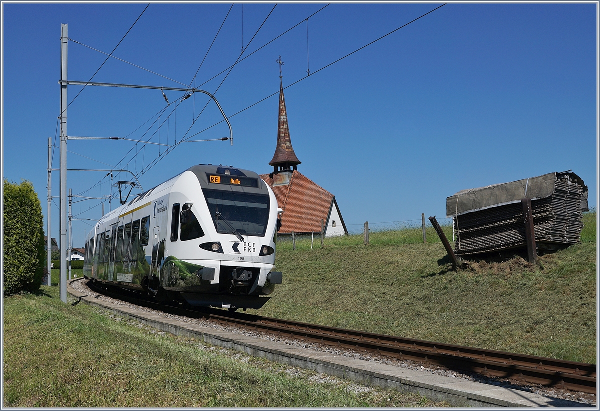 Der TPF RABe 527 198  BCF / FKB  (Kantonalbankwerbezug) erreicht Vaulruz, links im Bild die mittlerweile bekannte Kapelle von Vaulruz, rechts, die hier typischen an den Gleisböschung stehenden Bretterstapel. 

19. Mai 2020