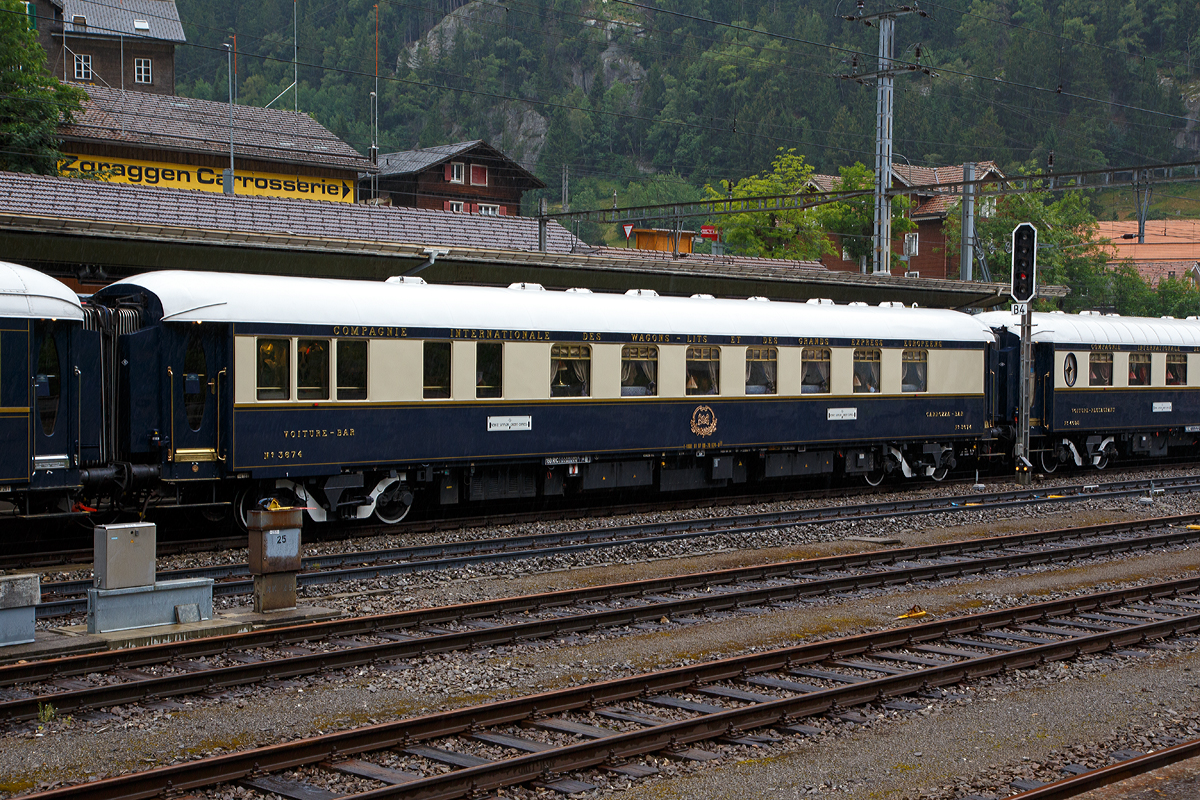 Der VSOE (Pullman) Barwagen Nr. 3674, Voiture-Bar / Carrozza-Bar N� 3674 (F- VSOE 61 87 09-70 674-0 [P]), ex SNCF WR 51 87 88-40 544-8, ex CIWL N� 3674, im Zugverbund vom Venice Simplon-Orient-Express am 02.08.2019 beim Halt in G�schenen (CH), leider bei Regen.

Der Barwagen wurde 1931 von EIC - Entreprises industrielles des Charentes in Aytr� (F) (EIC, heute zu Alstom geh�rend) gebaut und an die CIWL (Compagnie Internationale des Wagons-Lits) geliefert die ihn als Nr. 3674 bezeichnete, damals war es kein Pullman-Wagen. 

Seine Geschichte umfasst den Inlandsverkehr in Frankreich w�hrend der deutschen Besatzung im 2. Weltkrieg. nach dem Krieg den Sud-Express (S�d-Express) von Paris (Gare d’Austerlitz) �ber Orleans-Les Aubrais, Bordeaux (Saint-Jean), Bayonne und Biarritz nach Irun. Irun ist eine spanische Stadt nahe der spanisch-franz�sischen Grenze. Ab Irun ging es dann nur breitspurig oder meterspurig weiter. Er lief als WR 51 87 88-40 544-8 der SNCF. 

Ende der 1970er-Jahre wurde er an die VSOE verkauft. Die VSOE lie� den urspr�nglichen Speisewagen durch die Bremer Waggonbau GmbH (ehem. Hansa Waggonbau) 1982 zum Barwagen umbauen, Innengestaltung erfolgte nach G�rard Gallet. Der Wagen wurde sogar mit einem Fl�gel (Klavier) ausgestattet. Vermutlich erhielt er dort auch an einer Wagenseite (andere Wagenseite) ein typisches „Pullman-Fenster“ und wegen der eingebauten Bar an dieser Wagenseite auch einen l�ngeren fensterlosen Bereich. 

Zwischen 2003 und 2006 wurden die Wagen nochmals modernisiert und erhielten unter anderem moderne Drehgestelle von Bombardier sowie neue Klimaanlagen. Seitdem sind die Wagen nun f�r 160 km/h zugelassen.

TECHNISCHE DATEN:
Spurweite: 1.435 mm (Normalspur)
Anzahl der Achsen: 4 in zwei Drehgestellen
L�nge �ber Puffer: 23.452 mm
Drehzapfenabstand: 16.000 mm
Achsabstand im Drehgestell: 2.500 mm
Eigengewicht: 49 t
H�chstgeschwindigkeit: 160 km/h (urspr�nglich 140 km/h)
Sitzpl�tze: 36

Nach dem Dinner wird im Barwagen bei sanfter Musik Konversation betrieben. Der Pianist sitzt am Fl�gel. Es flie�t Champagner oder man nippt an Cocktails. Salzgeb�ck wird in Silbersch�lchen gereicht. Hier wird gelacht. Hier wurden und werden vielleicht immer noch Kontakte gekn�pft. Wer noch Hunger auf einen Snack versp�rt, kann sich Blinis mit Beluga-Kaviar anrichten lassen. Einige hundert Euro extra werden daf�r am Ende der Tour wohl auf der Rechnung stehen😉. Eine Fahrt im VSOE ist keine „All-inclusive-Reise“, auch wenn viele inclusive ist. 

Schlie�lich ist die Vorstellung vorbei und man zieht sich ins Abteil zur�ck. Vielleicht mit der passenden Bettlekt�re von Agatha Christie, um beim monotonen Klackadiklack der R�der besser einzuschlafen. War da nicht eben ein Schrei? Ist jemand aus dem schaukelnden Bett gefallen? Hat sich jemand im engen Abteil den Kopf gesto�en? Doch wieder ein Mord? Traum oder Wirklichkeit? Egal, sp�testens wenn man das Fr�hst�ck ans Bett serviert bekommt, werden alle erfahren, was in der Nacht im „K�nig der Z�ge“ geschehen ist.
