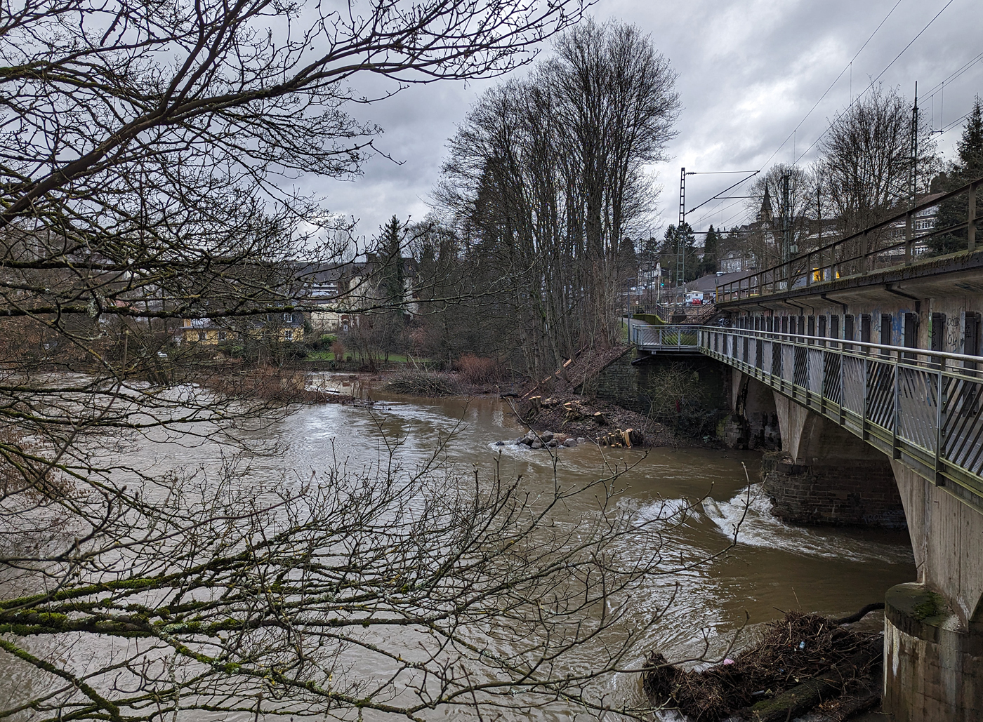 03.01.24	 Die vom Hochwasser beschädigte Eisenbahn-Siegbrücke mit Fußgängersteg in Kirchen (Sieg), der Siegstrecke (KBS 460), hier am 03.01.2024 (Bild mit Smartphone).

Die Bahnstrecke Siegen - Köln wird zwischen Kirchen und Betzdorf bis min. Ende nächster Woche gesperrt bleiben. Das hat die Deutsche Bahn Dienstagnachmittag mitgeteilt. Durch das Sieg-Hochwasser gibt es Schäden an einer Eisenbahnbrücke in Kirchen (kurz vorm Bahnhof). Aufgrund der Wetterlage und der weiterhin anhaltenden Regenfälle sei die Sperrung erforderlich, so die Bahn-Pressestelle. Die Züge des RE 9 können wegen der Streckensperrung nur bis Betzdorf fahren. Zwischen Siegen und Kirchen verkehrt die Regionalbahn 90 der Hessischen Landesbahn, leider aber nur wenige und teils mit „kleinen“ LINT 27 Triebwagen. Da wäre wahrhaft mehr möglich und man müsste den SEV nur zwischen Betzdorf und Kirchen betreiben. Aber aufgrund aktuell hoher Krankenstände kommt es auf der Linie RE 9 zudem zu Einschränkungen bis 12.01.2024.

Ich konnte mir den Schaden an einen Brückenpfeiler selbst anschauen (Bilder folgen) und finde die Streckensperrung richtig, darüber ließ ich auch keinen mit Personen besetzten Zug fahren. Was wäre wenn ein Zug darüber fährt und dadurch einbricht. Wer will das verantworten!!! 

Am 29.12.2023 wollten wir mit der Bahn nach Siegen, durch die Reiseauskunft unter www.bahn.de sahen wir bereits das durch wegen einer beschädigten Brücke zwischen Betzdorf (Sieg) und Kirchen (Sieg) ist eine Brücke beschädigt. Die Züge der Linie RE9 aus Richtung Köln enden und beginnen demnach in Betzdorf (Sieg). Die Züge der Linie RE 9 aus Richtung Siegen Hbf enden und beginnen demnach in Kirchen, diese waren aber nicht zu sehen. Bis dahin gab es keine Informationen zur Dauer der Sperrung. Somit fuhren wir mit dem Auto nach Kirchen und fuhren dann mit der RB 90 der HLB (Hessischen Landesbahn) in einem LINT 27, wie die Ölsardinen, nach Siegen Hbf. Auf der Rückfahrt um 14:31 Uhr (ab Siegen) mit RB 90 fuhren wir in einem LINT 41.

Am 30.12.2023 fuhren wir nochmal nach Siegen, das Wetter war besser und unser Ticket war noch gültig. Die Rückfahrt von Siegen machten wir um 13:31 Uhr mit dem HLB RB 90 nach Altenkirchen, dem HLB VT 261 einem LINT 41, dieser war der erste Zug der wieder durchfuhr und die Brücke in langsamer Fahrt befuhr. 

Später hieß es: Die Brücke zwischen Betzdorf und Kirchen ist nach wie vor beschädigt. Die Strecke ist aber wieder befahrbar. Die Züge fahren in dem betroffenen Streckenabschnitt langsamer. Reisende müssen mit Verzögerungen rechnen und sollten ihre Reiseverbindung kurz vor der Abfahrt des Zuges überprüfen.

Nun ist aber der betroffene Streckenabschnitt wieder gesperrt, die Brücke kann nicht mehr befahren werden.
 	google P6 pro						
