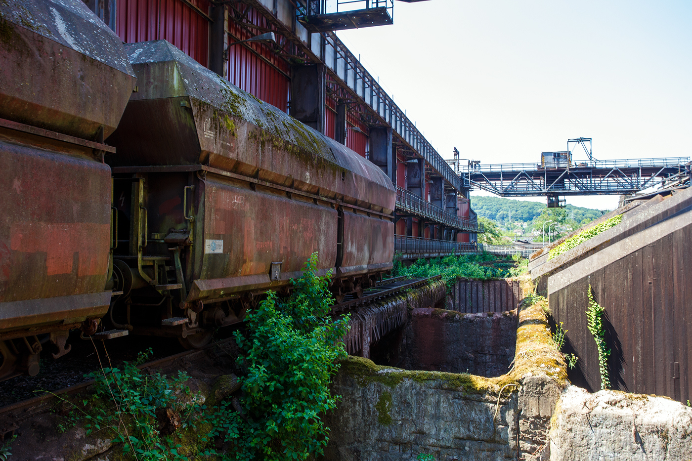 Am Tiefbunker (Rohstoffentladebunker der Sinterhalle) der Völklinger Hütte (ehem. Röchling'schen Eisen- und Stahlwerke GmbH in Völklingen/Saar, ab 1982 ARBED-Saarstahl GmbH, ab 1986 Saarstahl Völklingen GmbH) hinter der Normalspur-Denkmallok 34 stehen zwei vierachsige offene Schüttgutwagen (Sattelwagen) mit schlagartiger Schwerkraftentladung, hydraulischem Klappenverschlusssystem vermutlich der Gattung Falns (genaue Bezeichnungen kann man nicht erkennen), hier am 01 Juli 2025. Die Darstellung steht für den Rohstofftransport der Hütte zur Herstellung des Sintergutes. Die Lok war auch auf Bundesbahn Gleisen zugelassen. Heutiger Eigentümer der Lok und Wagen ist die Initiative Völklinger Hütte e.V..