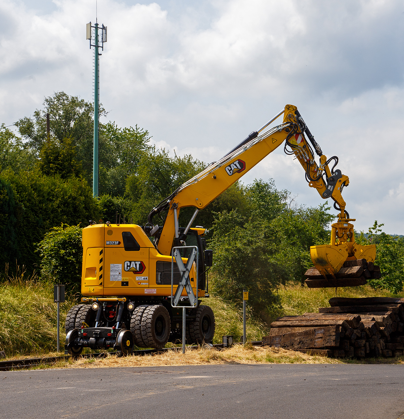Bei dem Rangierbahnhof der KSW (Kreisbahn Siegen-Wittgenstein) in Herdorf der Betriebsstätte FGE -Freien Grunder Eisenbahn (KSW NE447 / DB-Nr. 9275) wurden und wird der Unterbau der Gleise erneuert. Der Umbau erfolgt ohne große Einschränkungen des Tagesablaufes. 

Hier stapelt der Caterpillar CAT M323F Zweiwegebagger mit hydrostatischem Schienenradantrieb (Serien-Nr. RH600 189), Kleinwagen Nr. D-ZBM 99 80 9902 521-0 der ZEPPELIN Baumaschinen GmbH (Garching), wohl ein Mietbagger, am 26.06.2023 bei der Talbahnstraße Schellen.

Hier sieht man deutlich das der Fahrantrieb auf der Schiene, nicht wie bei vielen ZW-Baggern üblich, durch die Straßenreifen sondern mittels dem hydrostatischem Schienenradantrieb erfolgt (der Bagger bzw. das Straßenfahrwerk ist über die Schienen angehoben).

Der Zweiwegebagger wurde 2022 unter der Fabrik-Nr. CAT M323FERH600189 gebaut.