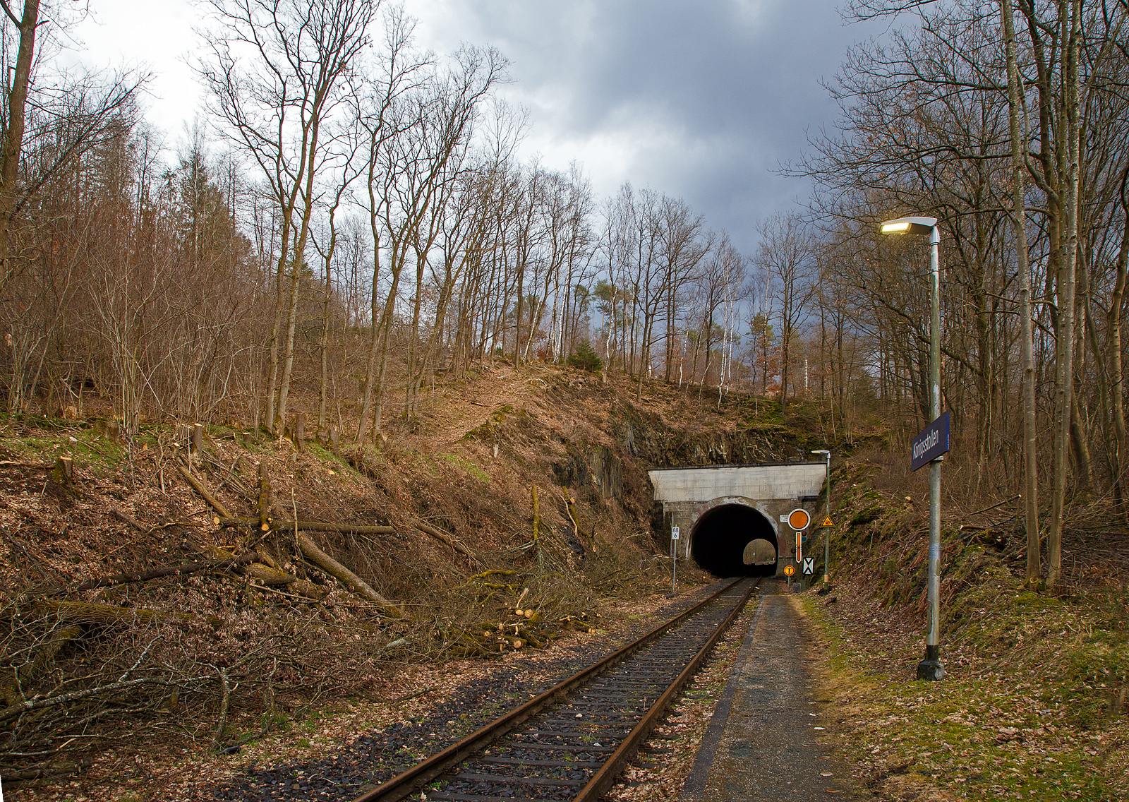 Der 137 m lange Herdorfer Tunnel hier am 18.03.2023 vom Hp K�nigsstollen gesehen. 

Davor das Vorsignal (f�rs Einfahrtsignal Bf Herdorf) Vr 0 - Halt erwarten! Die gelbe Scheibe mit wei�em Rand und gelbe Pfeil zeigt nach unten. Nachts leuchten auch die beiden gelben Lampen. Vor dem Signal die Ne 2 – Vorsignaltafel. Die Vorsignaltafel zeigt dem Lokf�hrer, da� das Signal, vor dem sie steht, Vorsignalfunktion hat, d.h., den Begriff eines nachfolgenden Hauptsignales vorank�ndigt. 
