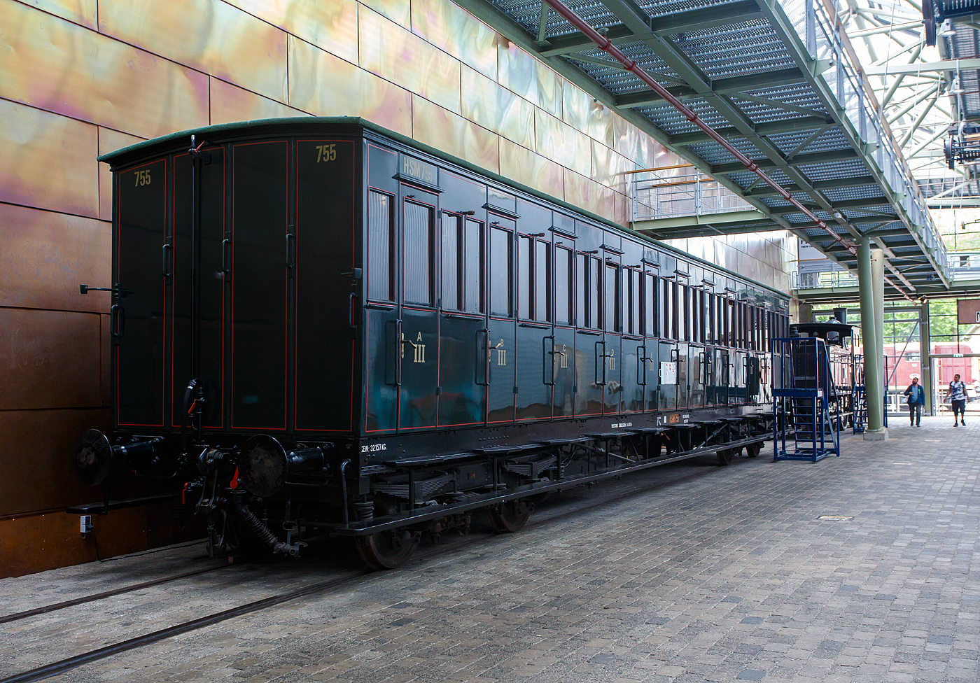 Der „viele T�ren Wagen“ ein 3. Klasse Abteilwagen mit Seitent�ren ohne Durchgangsm�glichkeit, HSM C 755 der damaligen HSM - Hollandsche IJzeren Spoorweg-Maatschappij (Holl�ndische Eisenbahn-Gesellschaft), ab 1922 NS C 5055, am 24. Juni 2025 im Het Spoorwegmuseum (bis 2005 NSM - Nederlands Spoorwegmuseum / Niederl�ndischen Eisenbahnmuseum) in Utrecht im ehemaligen Bahnhof Maliebaan.   

Ende des 19. Jahrhunderts hatten diese Wagen die Form eines langen, rechteckigen Kastens mit zahlreichen T�ren an den Seiten, die Zugang zu den Sitzabteilen boten. Eine lange, au�en angebrachte Stufe erm�glichte dem Schaffner den �bergang von einer T�r zur anderen. Die Sitzbereiche waren nur von au�en zug�nglich, einen Gang im Wageninneren gab es nicht. Anfang des 20. Jahrhunderts gab HSM 135 dieser Abteilwagen in Auftrag. Diese neue Wagengeneration war gr��er als die zuvor gebauten Wagen. Es gab 11 Abteile mit 10 Sitzpl�tzen f�r 110 Personen. Dies brachte den Wagen den Spitznamen „Schafstall“ ein. Diese Wagen waren im ganzen Land bis in die 1950er Jahre im Einsatz, als neue Wagen und Triebwagen die veralteten Abteilwagen ersetzten.

Der Wagen vom Typ C11 (Serie 707 - 766 III) wurde 1907 von der Nederlandsche Fabriek van Werktuigen en Spoorwegmaterieel in Amsterdam (ab 1929 Werkspoor N.V.) gebaut und an die HSM geliefert.

Bedingt durch den Ersten Weltkrieg wurde 1917 eine Zweckgemeinschaft der unabh�ngigen Eisenbahngesellschaften HSM beziehungsweise HIJSM (Hollandsche IJzeren Spoorweg-Maatschappij) und SS (Maatschappij tot Exploitatie van Staatsspoorwegen) gegr�ndet. Die beiden Gesellschaften errichteten einen gemeinsamen Bahnbetrieb, blieben aber rechtlich unabh�ngig. Dies �nderte sich 1938, als HIJSM und SS vollst�ndig fusionierten und in der AG NS (Nederlandse Spoorwegen, fr�her Nederlandsche Spoorwegen geschrieben) aufgingen. Das Rollmaterial wurde bereits 1921 zusammengelegt.

TECHNISCHE DATEN:
Spurweite: 1.435 mm (Normalspur)
Achsen: 4 (in 2 Drehgestellen)
L�nge �ber Puffer: 18.300 mm
Leergewicht: 32.157 kg
Sitzpl�tze: 110 in der 3. Klasse in 11 Abteilen � 10 Personen
H�chstgeschwindigkeit: 80 km/h