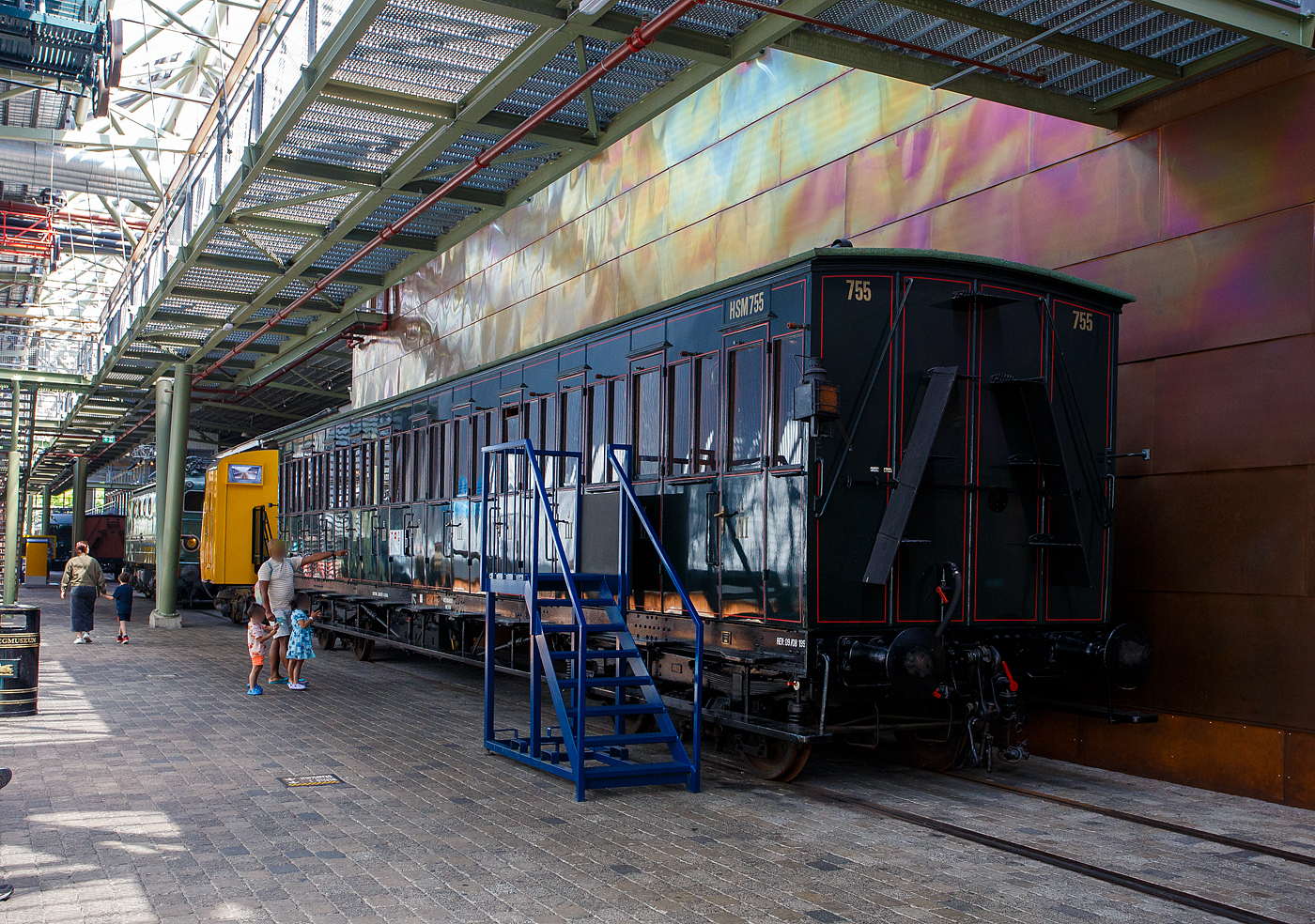 Der „viele T�ren Wagen“ ein 3. Klasse Abteilwagen mit Seitent�ren ohne Durchgangsm�glichkeit, HSM C 755 der damaligen HSM - Hollandsche IJzeren Spoorweg-Maatschappij (Holl�ndische Eisenbahn-Gesellschaft), ab 1922 NS C 5055, am 29 April 2025 im Het Spoorwegmuseum (bis 2005 NSM - Nederlands Spoorwegmuseum / Niederl�ndischen Eisenbahnmuseum) in Utrecht im ehemaligen Bahnhof Maliebaan.   

Ende des 19. Jahrhunderts hatten diese Wagen die Form eines langen, rechteckigen Kastens mit zahlreichen T�ren an den Seiten, die Zugang zu den Sitzabteilen boten. Eine lange, au�en angebrachte Stufe erm�glichte dem Schaffner den �bergang von einer T�r zur anderen. Die Sitzbereiche waren nur von au�en zug�nglich, einen Gang im Wageninneren gab es nicht. Anfang des 20. Jahrhunderts gab HSM 135 dieser Abteilwagen in Auftrag. Diese neue Wagengeneration war gr��er als die zuvor gebauten Wagen. Es gab 11 Abteile mit 10 Sitzpl�tzen f�r 110 Personen. Dies brachte den Wagen den Spitznamen „Schafstall“ ein. Diese Wagen waren im ganzen Land bis in die 1950er Jahre im Einsatz, als neue Wagen und Triebwagen die veralteten Abteilwagen ersetzten.

Der Wagen vom Typ C11 (Serie 707 - 766 III) wurde 1907 von der Nederlandsche Fabriek van Werktuigen en Spoorwegmaterieel in Amsterdam (ab 1929 Werkspoor N.V.) gebaut und an die HSM geliefert.

Bedingt durch den Ersten Weltkrieg wurde 1917 eine Zweckgemeinschaft der unabh�ngigen Eisenbahngesellschaften HSM beziehungsweise HIJSM (Hollandsche IJzeren Spoorweg-Maatschappij) und SS (Maatschappij tot Exploitatie van Staatsspoorwegen) gegr�ndet. Die beiden Gesellschaften errichteten einen gemeinsamen Bahnbetrieb, blieben aber rechtlich unabh�ngig. Dies �nderte sich 1938, als HIJSM und SS vollst�ndig fusionierten und in der AG NS (Nederlandse Spoorwegen, fr�her Nederlandsche Spoorwegen geschrieben) aufgingen. Das Rollmaterial wurde bereits 1921 zusammengelegt.

TECHNISCHE DATEN:
Spurweite: 1.435 mm (Normalspur)
Achsen: 4 (in 2 Drehgestellen)
L�nge �ber Puffer: 18.300 mm
Leergewicht: 32.157 kg
Sitzpl�tze: 110 in der 3. Klasse in 11 Abteilen � 10 Personen
H�chstgeschwindigkeit: 80 km/h

Quellen: Het Spoorwegmuseum, nmld.nl und Anschriften
Stand/�berarbeitung: September 2025
