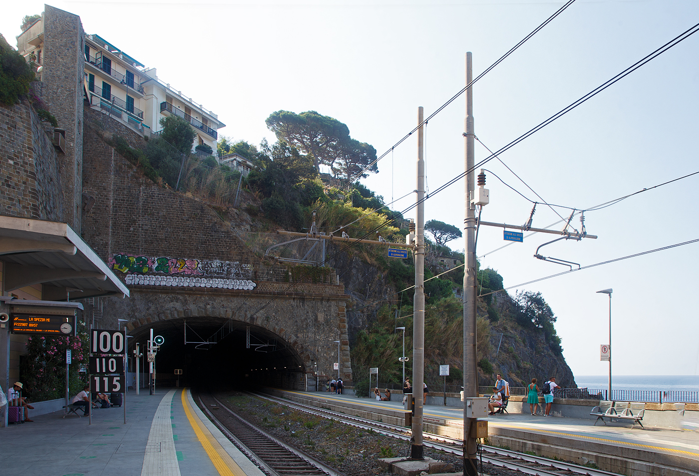 Der Cinque Terre Bahnhof Riomaggiore an der Bahnstrecke Pisa–Genua (RFI Strecke-Nr. 77 / KBS 31 La Spezia–Genua) am 22.07.2022. Hier mit Blick auf den südlichen Tunnel (in Richtung La Spezia), links vom Signal hinter den Tafeln ist der Eingang zu dem durch den Eisenbahntunnel verlaufenden Fußpfad zum Ortskern. Wiederrum links davon der hohe Lift (Aufzug), mit diesem kommt man auf den etliche Meter oben liegenden Weg. Der Lift ist aber in der Saison kostenpflichtig, er erspart einem aber etliche Treppenstufen oder einen gewaltigen Umweg.