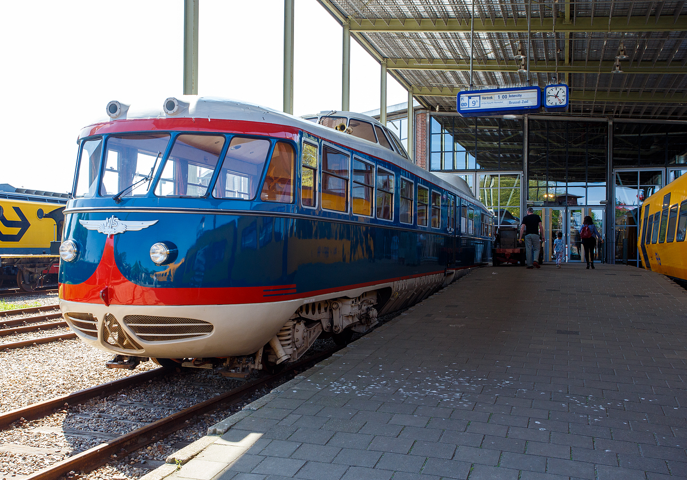 Der Dieseltriebwagen NS 20 „de Kameel“ / das Kamel bzw. VIP-Car, ein Dieseltriebwagen – Einzelstück und ehemaliger Inspektionstriebwagen, am 29 April 2025 im Het Spoorwegmuseum (bis 2005 NSM - Nederlands Spoorwegmuseum / Niederländischen Eisenbahnmuseum) in Utrecht-Maliebaan im Bahnhof Maliebaan. Ein sehr schönes Eisenbahnmuseum das zum Verweilen einlädt, leider fehlte uns dieses Mal die Zeit dafür. 

Der dieselelektrische Triebwagen wurde 1954 von Allan & Co´s Koninklijke Nederlandsche Fabrieken van Meubelen en Spoorwegmaterieel N.V. (Königlich Niederländische Möbel- und Eisenbahnausrüstungsfabrik) in Rotterdam gebaut und als Inspektionstriebwagen NS 20 an die NS - Nederlandse Spoorwegen (Niederländische Staatsbahnen) geliefert. Er diente für Inspektionsfahrten des NS-Vorstands.

Er technisch von den Triebwagen vom Typ DE 1 - Plan X „Blauwe Engel“ abgeleitet, nur der große Unterschied sind die Ausbuchtungen mit den Führerständen am Dach, die im bald den Spitznamen „Kamel“ einbrachten. 

1972 stand für den NS 20 eine Generalüberholung an. Zuvor durfte der Triebwagen in Toppop-Lackierung für die Musiksendung „Toppop“ durch die Niederlande touren. Im Rahmen der Überholung wurde der Triebwagen in Gelb umlackiert. Nach dieser Überholung stand der als „VIP-Wagen“ bekannte Triebwagen für Gruppenreisen zur Verfügung. Am 31. Januar 1991 fuhr der Wagen zum letzten Mal für die Öffentlichkeit, und am 8. März erfolgte die letzte Fahrt für die NS-Leitung. Anfang April fuhr die NS 20 mit eigenem Antrieb zur Lijnwerkplaats (Werkstatt) in Zwolle, um seine Generatoren als Ersatzteile zu spenden und sie der Reserve für die (inzwischen umgebauten und gelb lackierten) Blauwe-Engel-Züge hinzuzufügen. Diese Züge litten damals unter einem Mangel an Ersatzteilen.

Von diesem Zeitpunkt an war der NS 20 nicht mehr betriebsbereit und wurde zusammen mit einzelnen Dieselloks von der Lokomotive NS 2205 zum Eisenbahnmuseum transportiert und dort ausgestellt.

2005 wurde erneut ein Inspektionsfahrzeug benötigt, sodass eine Generalüberholung eingeleitet wurde, um das „Kamel“ wieder fahrbereit zu machen. Aus den ausgemusterten Plan X „Blaue Engel“ wurden Generatoren ausgebaut, zwei davon ausgewählt und nach Überholung wieder in den NS 20 eingebaut. Die Niederspannungsversorgung und die Küchenausstattung wurden in mehreren Bereichen für den modernen Einsatz modifiziert. Der in seiner originalen blauen Lackierung lackierte Triebwagen kehrte am 15. März 2008 mit eigener Kraft ins Museum zurück und wird dort betriebsbereit aufbewahrt.

Technik:
Auch die Frontpartie des Triebwagens anders als bei den DE 1 und 2. Das „Kamel“ ist 25.555 mm lang, somit fast 1,5 m kürzer als ein DE 1. Er hat eine Höhe von 4,145 Metern und eine Breite von 2,780 Metern. Das Gewicht des Fahrzeugs beträgt 58 Tonnen.

Der dieselelektrische Triebwagen verfügt über zwei Drehgestelle, die jeweils von zwei Elektromotoren angetrieben werden. Die elektrische Energie wird von zwei wassergekühlte AEC-6-Zylinder-Unterfur-Dieselmotoren Typ A220 (geliefert von Kemper & Van Twist), mit einer max. Leistung 132 kW (180 PS), Dauerleistung 107 kW (145 PS) mit gekoppeltem einem Generator von Smit vom Type G40/35 (147 kW) erzeugt, die jeweils zwei Heemaf-Fahrmotoren, Typ TM 51, antreiben. Die Dieselmotoren der DE 1 und 2 - Plan X „Blaue Engel“ hingegen haben durch einen Turbolader von Brown-Boveri eine höhere Dauerleistung von 165 kW (225 PS). Die maximale Drehzahl des Dieselmotors beträgt 1.250 U/min. Der Dieselmotor mit Generator, sowie Kompressor sind jeweils unter der Karosserie des Triebwagens montiert.

Der Triebwagen ist mit vier Heemaf-Fahrmotoren vom Typ TM 51 mit einer maximalen Leistung von 66 kW, 337 A bei 1.130 U/min und einer Dauerleistung von 49 kW, 248 A und 1.290 U/min ausgestattet. Die Energie liefert jeweils ein vom Dieselmotor angetriebener Generator für je 2 Motore. Kompressor und Hilfsgenerator werden über Keilriemen angetrieben. Diese befinden sich auf der anderen Seite des Hauptgenerators.

Der Radstand der Drehgestelle beträgt 3,0 Meter, der Mittenabstand zwischen den Drehgestellen 18,35 Meter. Der NS 20 ist für eine Höchstgeschwindigkeit von 125 km/h zugelassen. Der Raddurchmesser der Triebdrehgestelle, ist jedoch größer als bei den anderen Plan X-Garnituren, nämlich 1.250 mm. Die Drehgestelle sind jeweils mit zwei Heemaf-Fahrmotoren vom Typ TM 51 ausgestattet. Die Achsfolge ist somit Bo'Bo'. 

TECHNISCHE DATEN des DE 1 – 20 „de Kameel“:
Gebaute Anzahl: 1
Nummerierung: 20
Typ: dieselelektrischer Triebzug
Baujahr: 1954
Hersteller: Allan, Rotterdam
Spurweite: 1.435 mm (Normalspur)
Achsfolge: Bo'Bo' (4 Achsen in 2 Drehgestellen)
Länge über Kupplung: 25.550 mm
Drehzapfenabstand:
Achsanstand im Drehgestell: 3.000 mm
Breite: 2.780 mm
Höhe: 4.145 mm
Treibraddurchmesser: 1.250 mm (neu) 
Dieselmotor (2 Stück): wassergekühlte AEC-6-Zylinder-Unterfur-Dieselmotoren Typ A220
Motorhubraum: 11,31 Liter (Bohrung 130 mm x 142 mm Hub)
Motorleistung: 2 x 132 kW (180 PS)
Motordauerleistung: 2 x 107 kW (145 PS) bei 1.250 U/min
Generator (2 Stück): Smit G40/35 (147 kW)
E-Antriebsmotoren (4 Stück): Heemaf TM 51 max. 66 kW, 337 A bei 1.130 U/min
E-Motordauerleistung: 4 x 49 kW, 248 A und 1.290 U/min = 196 kW
Höchstgeschwindigkeit: 125 km/h
Eigengewicht: 58 t 

Quellen: Somda RailWiki.nl, Het Spoorwegmuseum, nmld.nl, Anschriften, Wikipedia (Niederlande)
Stand/Überarbeitung: September 2025
