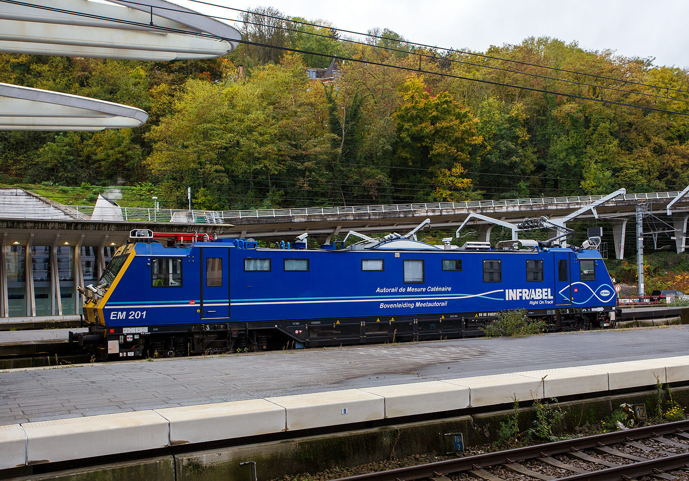 Der Oberleitungsmesstriebwagen (Autorail de Mesure Caténaire) EM 201 mit der UIC-Nummer 99 88 9165 201-3 B-INFBL, ein Geismar Eye Dragon VMB2214-IC der belgischen INFRABEL am 21 Oktober 2025 beim Bahnhof Lüttich-Guillemins (Gare de Liège-Guillemins). Kurzdrauf fuhr der Messtriebwagen die Schnellfahrstrecke in Richtung Ans bzw. Brüssel hinauf. 

Die Infrabel ist das 2005 aus der Privatisierung der Eisenbahn in Belgien hervorgegangene Infrastrukturunternehmen. Es ist vollständig im Eigentum des belgischen Staates und hat etwa 12.750 Mitarbeiter. Seine Aufgaben beinhalten die Instandhaltung sowie den Ausbau des 3602 km langen belgischen Schienennetzes, die Vergabe und Abrechnung von Fahrplantrassen sowie die Koordination des belgischen Zugverkehrs.

Die Infrabel hat 3 dieser Messtriebwagen vom Typ Geismar VMB2214-IC, die zwischen 2010 und 2014 gebaut wurden. Sie sind als Serie EM200 eingeordnet.

TECHNISCHE DATEN des Messtriebwagen:
Spurweite: 1.435 mm (Normalspur)
Achsfolge: Bo-Bo
Länge über Puffer: 21.240 mm
Drehzapfenabstand: 14.000 mm	
Achsabstand im Drehgestell: 2.500 mm
Gewicht: 89 t
min. Kurvenradius: R 90 m
