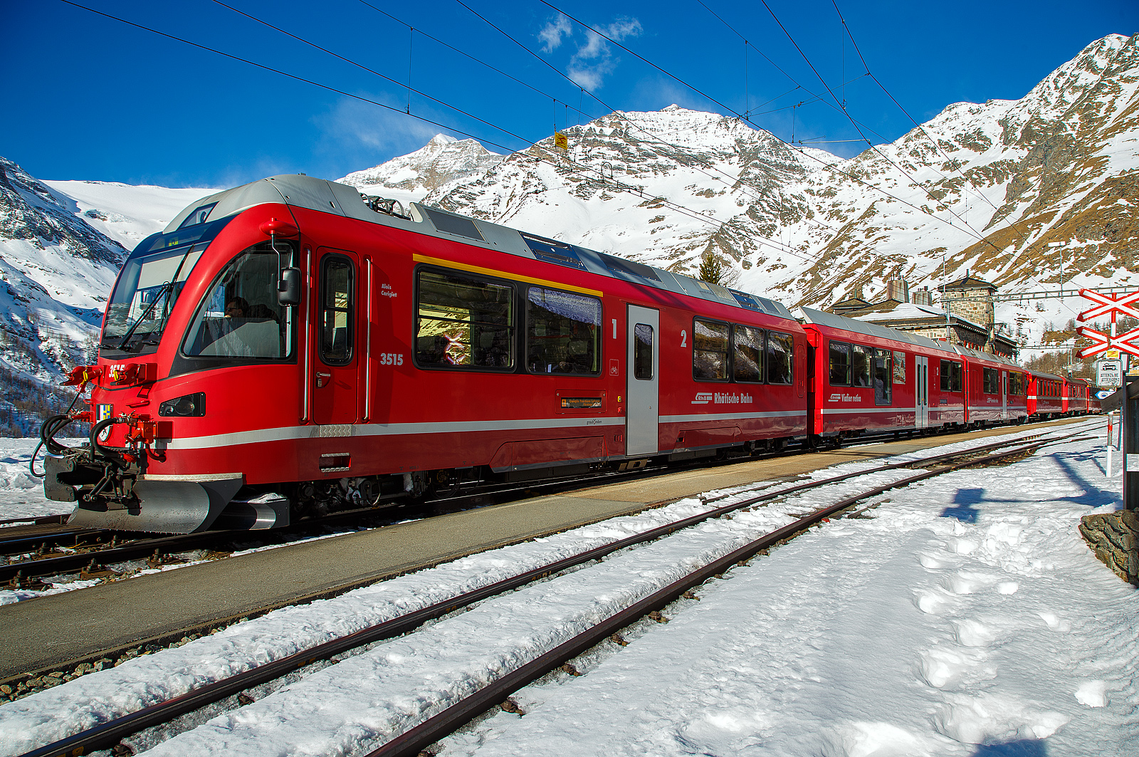 Der RhB ALLEGRA-Zweispannungstriebzug RhB ABe 8/12 3515  Alois Carigiet   mit 4 angehangenen Personenwagen, als RhB Regio nach Tirano hat am 20 Februar 2017 im Bahnhof Alp Gr�m (2.091 m �. M.).

Auf der Berninabahn sind die Triebz�ge das Haupttraktionsmittel und ziehen Regionalz�ge, den Bernina-Express sowie G�terz�ge. Die ABe 8/12 sind dreiteilige elektrische Triebz�ge, von denen die Rh�tische Bahn (RhB) bei Stadler Rail insgesamt 15 Einheiten beschafft hat. Die Triebz�ge werden von der RhB seit 2010 fahrplanm��ig eingesetzt. Die Z�ge werden als Allegra bezeichnet, was einer r�toromanischen Gru�formel entspricht.

Das Fahrzeugkonzept:
Anforderung
Die Grundvoraussetzung f�r den neuen Zweispannungstriebzug bestand darin, dass er freiz�gig auf dem ganzen Netz der RhB eingesetzt werden kann. Das hei�t, er muss auf der Berninalinie auf 70 ‰ Steigung gro�e Zugkr�fte aus�ben und trotzdem auf dem Stammnetz eine betriebliche H�chstgeschwindigkeit von 100 km/h erreichen k�nnen. Die Fahrzeugabmessungen werden ebenfalls von der Berninabahn mit den engen Kurvenradien von rund 42 m vorgegeben. Damit sind nur kurze Wagenk�sten m�glich, was bei den engen Platzverh�ltnissen eine ausgekl�gelte Verteilung der technischen Ausr�stung bedingt, sollen doch m�glichst viele Sitzpl�tze angeboten werden. Weitere Anforderungen waren komfortable Einstiege, ein eigenst�ndiges, modernes Design, Behindertengerechtigkeit sowie ein modernes Kundeninformationssystem.

Die technische L�sung
Das in der Ausschreibung geforderte Traktionsprogramm mit einer Anh�ngelast von 70 t auf 70 ‰ Steigung h�tte bei nur vier Triebachsen eine Achslast von �ber 14 t erfordert, was einen hohen Verschlei� an Rad und Schiene hervorrufen w�rde. Deshalb ist der Zug mit acht Triebachsen konzipiert. Somit k�nnen bei einer Achslast von nur 11 t sogar 140 t Anh�ngelast auf 70 ‰ Steigung bef�rdert werden, womit der Triebzug das maximale Zuggewicht ohne Vorspannlok oder ohne Doppeltraktion fahren kann.

Layout
Der Triebzug besteht aus zwei Endtriebwagen und einem Niederflurzwischenwagen. Um ein m�glichst gro�es Adh�sionsgewicht zu erreichen, wurde die gesamte Traktionsausr�stung in den beiden Endtriebwagen konzentriert. So konnten im Triebzug total 100 Sitzpl�tze geschaffen werden, wobei die 1. Klasse Abteile ein besonderes Fahrerlebnis bietet, nicht zuletzt der verglasten F�hrerstandr�ckwand wegen, die den Passagieren sozusagen am Lokf�hrer vorbei freie Sicht auf die Strecke erlaubt.

Die Fahrzeuggestaltung:
Aussendesign
Die Front des �ALLEGRA� ziert das B�ndner Kantonswappen. Es symbolisiert das Selbstbewusstsein und das Bekenntnis der RhB zum Kanton Graub�nden. Der untere Teil des F�hrerstands mit der zur Spitze laufenden Form und dem geraden, nach hinten gew�lbten F�hrerstandsfenster verleiht dem Fahrzeug Kraft und Stabilit�t, aber auch Dynamik. Die Lackierung der neuen Triebz�ge in RhB-Rot orientiert sich am bestehenden Rollmaterial. Der Bereich der Untersch�rzen ist neu in einer dunkelroten Farbe lackiert. Zusammen mit dem hellgrauen Trennstreifen wird so der Eindruck einer nach innen eingezogenen Sch�rze geschaffen. Die Fenster werden auf ihren Zwischenstreben mit einer dunklen Farbe verbunden und wirken so zusammengefasst. Trotz der kurzen Wagenl�nge und den verschiedenen Fensterh�hen wird das Erscheinungsbild dadurch wesentlich beruhigt, und die Zugkomposition, bestehend aus Triebzug und Verst�rkungswagen, bildet optisch eine Einheit.

Innendesign
Die RhB chauffiert ihre Kundschaft nicht nur durch eine spektakul�re Bergwelt, sondern auch durch die Kulturlandschaft Graub�ndens mit ihrem traditionellen Baustil. Das Innendesign des �ALLEGRA� nimmt diese Baukultur auf und interpretiert sie neu. Mit demselben Ansatz hat es die aktuelle B�ndner Architektur im �brigen zu Weltruhm gebracht. Um eine klare Struktur zu schaffen und dem Design eine gewisse Gro�z�gigkeit zu verleihen, wurden zwei Themen entwickelt, die durch das ganze Interieur f�hren: Als moderne Interpretation traditioneller Holzbauweise zieht sich die Oberfl�che �Trin� als Seitenwandverkleidung linear durch den ganzen Zug. Unterbrochen wird diese von den Apparatek�sten, die durch die Oberfl�che �Vals� als unregelm��ig geschichtete Monolithen erscheinen. Die Monitore der Fahrgastinformation wurden an den Abteilw�nden in mit schwarzem Glas belegte Horizontalstreifen integriert.

Echte Eisenbahnklasse
Um der teilweise niedrigen Raumh�he des ALLEGRA entgegenzuwirken, wird ein leicht h�her liegender Mittelstreifen von beiden Seiten horizontal angestrahlt. Dies sorgt f�r eine angenehme indirekte Beleuchtung. In der ersten Klasse akzentuieren kleine Halogen-Downlights diese sogar noch. Die Differenzierung der beiden Klassen erfolgt neben den �blichen Komfort-Parametern wie beispielsweise Sitzabstand durch die verschiedenen Bodenbel�ge und vor allem durch die Sitzbez�ge. Die Veloursstoffe im klassischen Karomuster verweisen auf die Tourismustradition, angefangen von den Golfhosen der englischen Upperclass bis zu den Wolldecken der Pferdekutschen. Das Muster der ersten Klasse verstr�mt im Zusammenspiel mit der pr�zisen Anmutung der Seitenwandverkleidung einen Hauch von Luxus, w�hrend in der zweiten Klasse die Blaut�ne ein Bild lebhafter Eleganz vermitteln.

Die Technik:
Die Ausschreibung der neuen Triebz�ge bestand aus einem anspruchsvollen Pflichtenheft. Das daraus resultierende Fahrzeugkonzept stellte f�r die Realisierung eine Herausforderung dar, wobei vor allem die Kombination aus hoher Antriebsleistung, Zweisystemf�higkeit, doppelter Bremsausr�stung, behindertengerechter Gestaltung bei beengten Platzverh�ltnissen durch Meterspur und die schwierigen Einsatzbedingungen der Berninalinie hervorzuheben ist.

Mechanischer Teil
Jeder der drei Wagen einer Einheit l�uft auf zwei zweiachsigen Drehgestellen und ist somit einzeln rollf�hig. Das Grundkonzept der Laufdrehgestelle wurde von den Bernina-Panoramawagen �bernommen. Die Wagen sind untereinander kurzgekuppelt, sie k�nnen nur in der Werkstatt getrennt werden. Die �berg�nge zwischen den Wagen sind durch Faltenb�lge gesch�tzt und von den Reisenden benutzbar. Der antriebslose Mittelwagen hat zwischen den Drehgestellen einen abgesenkten Wagenboden mit beidseitigen Niederflureinstiegen und Schiebetritten, um Personen mit eingeschr�nkter Mobilit�t einen barrierefreien Einstieg zu gew�hrleisten. Hinter den beiden F�hrerst�nden ist jeweils ein 1. Klasse Abteil vorhanden. Die Zwischenwand zum F�hrerstand ist verglast und bietet den Fahrg�sten einen Blick �ber die Schulter des mittig im F�hrerstand sitzenden Lokomotivf�hrers auf die Strecke. 

Eine Einheit bietet insgesamt 24 Sitzpl�tze in der ersten und 76 Sitzpl�tze in der zweiten Klasse, dazu kommen noch 14 Klappsitze und die Stellm�glichkeit f�r Rollst�hle.

Elektrischer Teil
Auf den Endwagen sitzt je ein Einholmstromabnehmer mit drei Schleifleisten f�r Gleichstrom, auf dem Mittelwagen einer f�r Wechselstrom. Im St�rungsfall und bei Rangierfahrten kann aber jeder Stromabnehmer unter beiden Spannungen benutzt werden. Alle drei Stromabnehmer sind �ber eine Hochspannungsdachleitung miteinander verbunden, das Stromsystem wird bei Befehl „Hauptschalter ein“ des Lokf�hrers vom Fahrzeug selbst�ndig erkannt. Beim Stromsystemwechsel muss der Lokomotivf�hrer im Stillstand oder w�hrend der Fahrt alle Stromabnehmer senken. Sobald wieder ein Stromabnehmer gehoben wird, erkennt das Fahrzeug das vorhandene Stromsystem. Bei Geschwindigkeiten �ber 15 km/h wird mit gesenkten Stromabnehmern leicht elektrisch gebremst. Die dabei gewonnene Energie dient zur unterbrechungsfreien Versorgung der Hilfsbetriebe (St�tzbremsbetrieb).

Die ABe 8/12 verf�gen �ber eine Fernsteuerung zur Bildung von Pendelz�gen. Viele Komponenten sind technisch eng verwandt mit den gleichzeitig entwickelten RABe 4/12 „NExT“ der RBS).

Eine kraftvolle Traktionsausr�stung
Der Triebzug soll (wie bereits erw�hnt) ein breites Spektrum von Einsatzm�glichkeiten abdecken, was die Ingenieure in vielerlei Hinsicht vor H�rden stellte. Um den Anforderungen in Bezug auf die Beschleunigungszeiten mit verschiedenen Anh�ngelasten in unterschiedlichen Steigungen gerecht werden zu k�nnen, muss der Triebzug eine maximale Zugkraft von rund 280 kN auf die Schiene bringen k�nnen. Bei einer Dauerleistung von 2.320 kW kann die maximale Leistung auf 2.800 kW erh�ht werden.

F�r den Antrieb kommen vier wassergek�hlte Traktionsstromrichter aus der BORDLINE�-Familie der Firma ABB zum Einsatz. Auch die �lgek�hlten Transformatoren, untergebracht in einem robusten Stahlkessel f�r Unterflurmontage, kommen von ABB S�cheron. Im Transformatorkessel sind zus�tzlich die zwei Netzdrosseln f�r den Betrieb am Gleichstromnetz montiert. Die gemeinsame K�hleinheit f�r Stromrichter und Transformator sind jeweils in der Mitte der Endwagen auf dem Dach platziert. Bei den Fahrmotoren handelt es sich um vierpolige, fremdbel�ftete Asynchronmotoren von TSA. Dieselben Fahrmotoren sind auch bei den neuen Triebz�gen f�r die Gesellschaften BDWM, RBS und LEB im Einsatz.

Bremsen und Bedienung
Die Triebz�ge verf�gen wegen der unterschiedlichen Bremsausr�stung der Bestandsfahrzeuge �ber mehrere Bremssysteme. Als Betriebsbremse wirkt vorwiegend die elektrodynamische Rekuperationsbremse, die sowohl im Wechselstrom- als auch im Gleichstrombetrieb die Bremsenergie ins Fahrleitungsnetz zur�ckspeist. F�r den Fall, dass das Fahrleitungsnetz nicht aufnahmef�hig ist, sind zus�tzlich Bremswiderst�nde vorhanden. Die elektrische Bremse kann vom Lokomotivf�hrer mit dem Zugkraft/Bremskraft-Hebel oder �ber die Geschwindigkeitsregelung angesteuert werden. Zur Begrenzung der Pufferkraft ist die Bremskraft der elektrischen Bremse auf 130 kN begrenzt.

Die indirekt wirkende Druckluftbremse steuert innerhalb des Triebzuges in jedem der drei Wagen �ber ein Steuerventil die Klotzbremse an. Die Drucklufthauptleitung ist an den Stirnseiten �ber je zwei Schlauchkupplungen herausgef�hrt. Damit k�nnen entsprechend ausgestattete Wagen, zum Beispiel Albula-Gliederz�ge, mit Druckluft gebremst werden.

Die Vakuumbremse ist die gebr�uchlichste Zugbremse bei der RhB. �ber den Zugkraft/Bremskraft-Hebel werden die Drucklufthauptleitung f�r den Triebzug und die Vakuumhauptleitung f�r die Anh�ngewagen gemeinsam gesteuert. Weil die druckluftgebremsten ABe 8/12 normalerweise mit vakuumgebremsten Z�gen verkehren, erfolgt die Bremsbedienung wie bei einer RhB-Lokomotive mit getrennter Bedienung der elektrischen und pneumatischen Bremse. Damit der Triebzug bei einer Unterbrechung der Vakuumhauptleitung, zum Beispiel beim Bet�tigen der Notbremse im Zug oder durch eine Zugtrennung, trotzdem bremst, ist zus�tzlich ein Vakuumsteuerventil vorhanden, das auf die Druckluftbremse wirkt.

Die direkt wirkende Rangierbremse kann die Geschwindigkeit des Triebzuges unabh�ngig von der Zugsbremse verringern. Die Bedienung erfolgt �ber den Rangierbremshebel. Eine Federspeicherbremse dient als unersch�pfliche Feststellbremse. Sie wirkt auf alle Trieb- und zwei Laufrads�tze. F�r den Einsatz auf der Berninabahn sind die Z�ge mit einer Magnetschienenbremse ausgestattet. Sie wirkt bei einer Schnellbremsung oder wenn sich der Zugkraft/Bremskraft-Hebel und der Zugsbremshebel in der hintersten Stellung befinden.

Die Allegra-Z�ge verf�gen �ber eine sehr pr�zise arbeitende Geschwindigkeitsregelung mit getrennter Vorgabe von Sollgeschwindigkeit und Zugkraft/Bremskraft. Mit dem Zugkraft/Bremskraft-Hebel stellt der Lokomotivf�hrer die Beschleunigung ein, mit der die Sollgeschwindigkeit erreicht werden soll. Unabh�ngig von der Streckenneigung wird diese Geschwindigkeit gehalten, wenn die eingestellte Zug- oder Bremskraft dazu ausreicht.

Der F�hrerraum
Mit den neuen Triebz�gen hat auch ein neues F�hrerraumkonzept bei der RhB Einzug gehalten. Erstmals wurde das bisher stets rechts angeordnete F�hrerpult in der Mitte angeordnet. Der helle F�hrerraum mit dem klar strukturieren F�hrerpult besticht durch seine Gro�z�gigkeit und die guten Sichtverh�ltnisse, die insbesondere f�r die Stadtstrecken in Chur und der Berninalinie wichtig sind.

Der Komfort im �ALLEGRA�
Klimaanlage
Die Klimatisierung des �ALLEGRA� erfolgt �ber Kompakt-Klimager�te auf dem Fahrzeugdach. Jeder Wagen ist mit einem separaten Klimager�t f�r den Fahrgastraum ausger�stet. Der F�hrerstand verf�gt dar�ber hinaus �ber ein eigenst�ndiges Klimager�t, welches ebenfalls auf dem Dach platziert ist.

Die Fenster
Die Seitenscheiben sind als Doppelverglasung aus w�rmebeschichtetem Sicherheitsglas ausgef�hrt. Die gew�hlte Kombination bietet eine optimale Lichtdurchl�ssigkeit bei gleichzeitig hoher UV- und W�rmeisolation. Manuell zu �ffnende �bersetzfenster bieten den Fahrg�sten eine gute M�glichkeit zum Fotografieren. Diese �bersetzfenster sind konzeptionell auch als �Redundanz� beim Ausfall einer Klimaanlage vorgesehen, indem im Sommer durch die Fahrtluft die Temperatur im Innenraum im zumutbaren Rahmen gehalten werden kann.

Steckdosen
Viele Fahrg�ste m�chten ihre Laptops und Handys w�hrend einer Zugfahrt aufladen k�nnen. Zu diesem Zweck sind alle Seitenwandtische des �ALLEGRA� mit je zwei 230V AC Steckdosen ausger�stet.

Toilette
Die Toilette ist im Niederflurteil des Mittelwagens platziert und vollst�ndig behinderten- und rollstuhlgerecht gestaltet. Im Weiteren sind eine LED-Spotbeleuchtung, ein separater Konvektionsheizk�rper und ein ausklappbarer Wickeltisch vorhanden. Der WC-Korpus entspricht dem bereits vom Glacier Express bekannten und bew�hrten WC-System.

Das Kundeninformationssystem
Gro�z�gige 24-Zoll-TFT-Monitore in den Fahrgastr�umen und Matrixanzeigen im Frontbereich sowie LED-Anzeigen an den Seiten informieren die Fahrg�ste �ber die n�chste Station, die Endstation, die Uhrzeit und weitere Informationen wie Versp�tungen, Anschlussm�glichkeiten und Sondermeldungen. Auch Bilder k�nnen mittels Playlisten �ber die Monitore angezeigt werden, sodass beispielsweise eine Reisegesellschaft den �ALLEGRA� mieten und entsprechend angeschrieben auf die Reise schicken kann, w�hrendem Reiseinformationen z.B. �ber die Berninalinie, den Fahrg�sten auf den TFT angezeigt werden.

Spezielle Einrichtungen f�r mobilit�tseingeschr�nkte Personen
Diesem Aspekt wurde vor allem im Niederflurteil des Mittelwagens Rechnung getragen. Durch die gro�en, doppelfl�geligen Einstiegst�ren mit einer lichten Weite von 1.200 mm und Schiebetritten zur Spalt�berbr�ckung wird der Einstieg am Normperron stufen- und spaltlos erm�glicht. Im Niederflurteil k�nnen f�r Individualreisende zwei Rollstuhlpl�tze reserviert werden. Im Bereich der Panoramafenster ist ein attraktives Behindertenabteil eingerichtet, das durch Hochklappen der Sitzpolster Platz f�r einen Rollstuhl mit Begleitperson bietet. Elektrisch bet�tigte Fensterrollos und tiefer angebrachte TFT-Monitore bieten zus�tzlichen Fahrkomfort. Bei Nichtgebrauch entsteht durch Herunterklappen der Sitzpolster eine vollwertige 2. Klasse-Sitzgruppe. Im Rahmen von Sonderfahrten und Gruppenreisen ist es �berdies m�glich, im gesamten Niederflurbereich f�r bis zu zehn Personen im Rollstuhl Pl�tze anzubieten.

Einsatz
Nach Abschluss der Typenpr�fungen erfolgen die Kundenabnahmen der ersten �ALLEGRA�-Triebz�ge ab April 2010 durch die RhB. Nach dem Fahrplanwechsel im Mai 2010 wurde der Regelbetrieb mit den neuen Triebz�gen aufgenommen. Die weitere Fahrzeugablieferung erfolgt anschlie�end monatlich ab Juli 2010 und wurde mit der Netzabnahme des letzten Zweispannungstriebzuges 3515 im April 2011 abgeschlossen sein. Anschlie�end wurden die f�nf vierteiligen Stammnetztriebz�ge ausgeliefert.

Geschwindigkeitsrekord auf Meterspur:
Anl�sslich der Abschiedsfahrt f�r den scheidenden Direktor wurde am 20. Dezember 2010 mit dem �ALLEGRA� 3502 im Vereinatunnel eine Geschwindigkeit von 145 km/h erreicht. Zuvor war bei den Inbetriebsetzungsfahrten am 5. Dezember 2009 139 km/h erreicht worden. 

TECHNISCHE DATEN der RhB ABe 8/12: 
Nummerierung:  3501–3515
Anzahl:  15
Hersteller:  Stadler Rail
Baujahre:  2009–2010
Achsformel:  Bo’Bo’+2’2’+Bo’Bo’
Spurweite:  1.000 mm (Meterspur)
L�nge �ber Puffer:  49.500 mm
H�he:  3.800 mm
Breite:  2.650 mm
Leergewicht:  106 t
H�chstgeschwindigkeit:  100 km/h
Max. Leistung (am Rad): 2.800 kW bei AC / 2.400 kW bei DC 
Dauerleistung: 2 320 kW
Anfahrzugkraft:  280 kN
Treibraddurchmesser:  810 mm (neu) / 740 mm (abgenutzt)
Laufraddurchmesser:  685 mm  (neu) / 635 mm (abgenutzt)
Achsabstand im Motordrehgestell: 2.000 mm
Achsabstand im Laufdrehgestell: 1.800 mm
Stromsystem:  11 kV 16.7 Hz AC und 1 kV DC
Anzahl der Fahrmotoren:  8
Sitzpl�tze:  1. Klasse: 24 / 2. Klasse: 76 + 14 Klappsitze
Fu�bodenh�he:  480 mm Niederflur / 1.050 mm Hochflur
Etiegbreite: 850 mm (Endwagen) und 1.200 mm (Mittelwagen)
Niederfluranteil:  22 % 

Die Anh�ngelastentabelle der ABe 8/12:
35 ‰  Albulalinie       60 km/h 	245 t
45 ‰  Davoserlinie 	55 km/h 	185 t
60 ‰  Arosalinie 	35 km/h 	200 t
70 ‰  Berninalinie 	40 km/h 	140 t


Quelle: RhB Medientext vom 1. Mai 2010, Wikipedia (de), Stadler Rail
