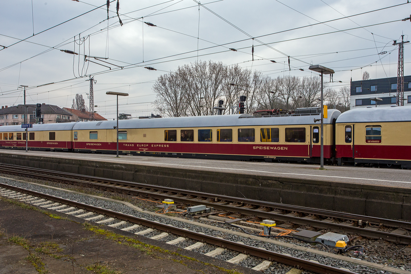 Der TEE-Speisewagen D-AKE 56 80 88-95 001-5 der Gattung WRmz 135.0, der AKE-Eisenbahntouristik – Jörg Petry e.K. (Gerolstein) am 10 März 2024 im Hbf Osnabrück, eingereiht im Zugverband in den 1. Klasse-Sonderzug AKE RHEINGOLD (vom 10 bis 14 März 2024) von Koblenz via Hamburg, Bremen, Husum und Westerland auf Sylt nach List auf Sylt.

Die zunehmende Elektrifizierung des Streckennetzes der Deutschen Bundesbahn erlaubte in der zweiten Hälfte der sechziger Jahre auch in Deutschland die Entwicklung vollelektrischer Speisewagen. Die Schweizerischen Bundesbahnen beschafften derartige Fahrzeuge zu diesem Zeitpunkt bereits seit 25 Jahren.

Dieser Speisewagen wurde 1969 von der Waggonfabrik Orenstein & Koppel in Berlin-Spandau als einer von sechs Prototypen gebaut und im November 1969 mit der Wagennummer 61 80 88-73 304-2 in Dienst gestellt. Ihre zulässige Höchstgeschwindigkeit betrug damals 160 km/h, in der Küche erhielten sie neben einem E-Herd mit acht Kochplatten erstmals eine Geschirrspülmaschine. Das im Vergleich zu den bislang beschafften Speisewagen auffälligste äußere Kennzeichen war der Dachstromabnehmer. Er konnte nur im Stillstand des Wagens an die Oberleitung angelegt werden und sicherte damit die Stromversorgung der Küche während der Abstellung, bei der Zugvorbereitung und im Betriebseinsatz beim Lokwechsel: Damals gab es noch keine IC-Steuerwagen und bei der Änderung der Fahrtrichtung wechselte stets die Lok, beispielsweise im großen Kopfbahnhof Frankfurt/M. Der Stromabnehmer senkte sich automatisch, wenn sich der Wagen in Bewegung setzte oder über die Heizleitung wieder Spannung erhielt.

Die Konzeption der Energieversorgungsanlage ließ nur Einsätze in den Bahnstromnetzen von Deutschland, Österreich und der Schweiz zu. Beheimatet war der Wagen von Anbeginn im Hamburg, daran sollte sich auch über 20 Jahre nichts ändern. Er lief zunächst im Hispania-Express zwischen Hamburg und Genf, nach der Beseitigung der bei Prototypen üblichen anfänglichen Störungen folgten ab 1971 Einsätze in den TEE „Helvetia“ nach Zürich und „Blauer Enzian“ nach Klagenfurt.

1977 wurde der Wagen durch Umbauten an den Drehgestellen und das Anbringen von Schlingerdämpfern für eine Höchstgeschwindigkeit von 200 km/h ertüchtigt. Ab 1979 oblag ihm die Bewirtschaftung von IC- und später EC-Zügen vorwiegend in die Schweiz mit weiterhin klangvollen und bekannten Namen wie „Rheinpfeil“, „Helvetia“, „Mont-Blanc“ und „Tiziano“. Eine komplette Vollaufarbeitung erfolgte im Herbst 1985, dabei erhielt er u. a. neue Fenster und Sitzbezüge im bekannten orange/braun/beige-gestreiften Muster. Ab Anfang der 90er-Jahre war dieser Wagen in Basel beheimatet und kam vorwiegend mit IC-Zügen in seine „Geburtsstadt“ Berlin.

1999 wurde eine neue Küche eingebaut, außerdem wurden die Sitzbezüge und Teppiche erneuert. Danach fand sich für das Fahrzeug bis 2006 ein neues Einsatzgebiet als Speisewagen für die DB AutoZug GmbH.

Nach seiner Ausmusterung bei der DB wurde der Wagen von der AKE-Eisenbahntouristik erworben und ist nach einer weiteren Modernisierung fester Bestandteil in den TEE „Rheingold“-Sonderzügen.

Quelle: AKE-Rheingold

TECHNISCHE DATEN:
Länge über Puffer: 27.500 mm
Wagenkastenlänge: 27.200 mm
Wagenkastenbreite: 2.805 mm
Höhe über Schienenoberkante: 4.050 mm
Drehzapfenabstand: 19.500 mm
Achsstand im Drehgestell: 2.500 mm
Drehgestellbauart: Minden-Deutz 367
Leergewicht: 49 t
Höchstgeschwindigkeit: 200 km/h (bei Ablieferung 160 km/h bis 1977)
Sitzplätze: 48  (Jeweils sieben Tischen für zwei bzw. vier Persone).
Heizungsbauart: Klimae
Energieversorgung: Zentrale Energieversorgung aus der Zugsammelschiene, im Stillstand/ohne Lok über Dachstromabnehmer

Allgemeines zu den TEE/IC-Wagen
Mit der Umstellung des bisher zweiklassigen Rheingold-Express-Zuges wollte die DB neuestes Wagenmaterial einsetzen. Vorgesehen waren reine Erste-Klasse-Wagen in mehreren Bauarten (die als Rheingold-Wagen bekannt sind). Diese Wagen wurden unter Leitung von Abteilungspräsident Adolf Mielich, Leiter der Wagenbau- und Einkaufsabteilung des Bundesbahnzentralamts in Minden, konstruiert. Sie besitzen somit die gleichen Hauptmaße wie die m-Bauart, waren damit 26,4 Meter lang und 2.825 Millimeter breit. Die durch Gummiwülste geschützten Wagenübergänge waren nach neuen UIC-Anforderungen aber nunmehr durch zweiflügelige Schiebetüren gesichert. Untergestell und Wagenkasten sind wiederum aus verschiedenen Profilblechen der Stahlgüte St52 zusammengeschweißt. Ebenso von den UIC-X-Wagen übernommen wurden die Drehfalttüren und die Dachform. Jedoch waren die neuen Wagen klimatisiert. Der Bereich unterhalb des Wagenbodens wurde durch eine Schürze ähnlich den Schürzenwagen geschützt.

Bei den Drehgestellen kam wie bei den m-Wagen die Bauart Minden-Deutz zum Einsatz, die aber für höhere Geschwindigkeiten ausgelegt wurde (MD36). Die Wagen waren zuerst für 160 km/h zugelassen und mit Klotzbremsen und einer Magnetschienenbremse ausgerüstet. Nachdem die Klotzbremsen durch Scheibenbremsen ersetzt wurden, konnte die Höchstgeschwindigkeit auf 200 km/h erhöht werden.

Bauart Helvetia
Zum Jahresfahrplan 1965 wurden weitere F-Züge aufgewertet und in TEE-Züge umgewandelt (z. B. „Blauer Enzian“). Der TEE „Helvetia“ wurde von Triebzügen der Baureihe VT 11.5 auf einen Lok-Wagen-Zug umgestellt. Dafür mussten weitere Wagen angeschafft werden. Diese unterscheiden sich von den vorangegangenen (Rheingold-Wagen) durch das nun zur Ausführung gekommene Steildach (bei den Sitzwagen erst ab dem Jahr 1967). Das bedeutet, die Wagendächer waren bis zum Wagenende gerade durchgezogen. Sonst glichen die Wagen einander. Bis 1975 wurden 266 Avmz111 und 99 Apmz121 gebaut. Einige Avmz111 der letzten Serie hatten Schwenkschiebetüren, die allerdings in den 1990er Jahren durch die normalen Drehfalttüren ersetzt wurden.
