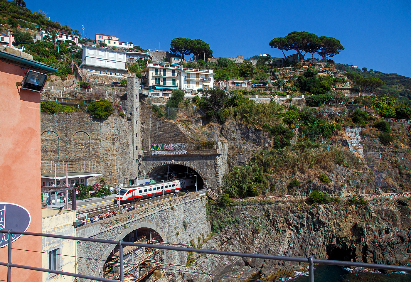 Der Trenitalia Frecciabianca ETR 460.23 (Pendolino) fährt am 22.07.2022 durch den Bahnhof Riomaggiore (Cinque Terre) in Richtung La Spezia bzw. Pisa. 

Riomaggiore ist das südlichste der fünf Dörfer der Cinque Terre (Fünf Ortschaften) und verfügt, wie die anderen Dörfer, über einn Bahnhof an der Bahnstrecke Pisa–Genua, die den Ort mit den Nachbardörfern und mit La Spezia und Levanto verbindet. Der Bahnhof von Riomaggiore liegt am nordwestlichen Ortsrand weitgehend im Tunnel, nur drei bis vier Wagenlängen befinden sich unter freiem Himmel. Der Ortskern wird mittels, des durch den im Bild zusehenden Eisenbahntunnel verlaufenden abgegrenzten Fußpfades angebunden (Eingang kurz vor dem Zugende).Zudem sieht man hinten auch den hohen Lift (Aufzug), mit diesem kommt man auf den etliche Meter oben liegenden Weg. Der Lift ist aber in der Saison kostenpflichtig, aber er erspart etliche Treppenstufen oder einen gewaltigen Umweg.

Der ETR.460 (von italienisch ElettroTreno Rapido) oder umgangssprachlich Pendolino ist ein von Trenitalia betriebener italienischer Neigezug von Fiat Ferroviaria. Noch während die Produktion der ETR.450 im Gange war, wurde mit dem Bau der zweiten Generation von Pendolino-Zügen begonnen. Die 10 Züge wurden 1991 von Italienischen Staatsbahn (FS) bestellt. Der fahrplanmäßige Einsatz, der 9-teiligen Züge, begann 1995 auf der Strecke Rom–Mailand–Venedig. Weiter verkehrten Züge von Rom aus auch nach den Städten Bari, Bergamo, Bozen, Lecce, Reggio Calabria und Savona. Seit 2013 gehören die Züge zur Zuggattung Frecciabianca und bedienen von Rom aus Genua, Ravenna (und bis 2020 Reggio Calabria), sowie seit 2021 die Verbindung Venedig–Lecce über die Adriabahn.