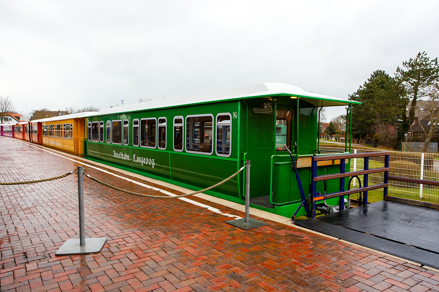 Der vierachsige meterspurige Personenwagen 94/04 (Sitzwagen) der Gattung KB4 der Inselbahn Langeoog vom Eigenbetrieb der Schiffahrt der Inselgemeinde Langeoog am 13 Februar 2024 im Insel Bahnhof Langeoog (im Ort Langeoog).

Der Wagen wurde von der Bremer Waggonbau GmbH 1994/95  gebaut. Die Serie für Inselbahn Langeoog bestand aus 10 Personen- und 1 Containertragwagen. Mit diesen elf neu gebauten Wagen für die Inselbahn Langeoog endete die Geschichte der Bremer Waggonbau GmbH, sie ging anschließend in Insolvenz, aber nicht wegen der Inselbahn. 

TECHNISCHE DATEN der Sitzwagen 94/01 bis 94/08:
Spurweite: 1.000 mm
Achsanzahl: 4
Länge über Kupplung: 15.000 mm
Länge über Rahmen: 14.200 mm
Drehzapfenanstand: 8.970 mm
Achsabstand im Drehgestell: 1.800 mm
Laufraddurchmesser: 650mm (neu)
Breite: 2.550 mm
Höchstgeschwindigkeit: 25 km/h
Eigengewicht:  14.500 kg
Sitzplätze: 62

Für interessierte im Anhang eine etwas längere Beschreibung der Wagen:
Die Fahrzeugrahmen bestehen aus handelsüblichen U- und Winkelprofilen der Stahlgüte St 52-3, die miteinander verschweißt wurden. Die Wagenkästen, die mit Edelstahlblechen außen verkleidet sind, wurden nicht als Ganzes auf den Fahrzeugrahmen aufgesetzt, sondern entstanden in einzelnen Segmenten, die dann auf den Rahmen aufgesetzt und angepasst wurden, dann wurden sie auf diesen verschweißt. Zusammen mit dem ebenfalls aus Edelstahl bestehenden Dach bilden Fahrzeugrahmen und Aufbau eine sehr verwindungssteife Einheit. Der Grund der Verwendung von Edelstahl ist die salzhaltige Luft an der Nordsee, normaler Baustahl oxidierend (rostet) hier viel schneller. Bevor die ersten Lackierarbeiten ausgeführt wurden, sind die auch aus Edelstahl bestehenden Bühnengeländer montiert worden, die gleichzeitig als Regenfallrohre für das Wagendach dienen. Die Edelstahlbleche des Wagenkastens wurden von innen und außen gesandstrahlt,  um eine bessere Haftung für den Farbaufbau zu erreichen.

Besonderer Wert wurde auf den Korrosionsschutz der Wagen gelegt, damit sie möglichst lange dem rauen Nordseeklima widerstehen können, Zur Minderung des Körperschalls wurde im Wageninnern Antidröhn-Masse aufgetragen und der Wagen mit Isolierungsmaterial ausgekleidet. Der Fußboden ist zweischalig ausgeführt und kann deshalb einen breiten Kabelkanal aufnehmen, in dem alle wichtigen Kabelverbindungen zusammengefasst sind. Die elektrische Ausrüstung der Wagen lieferte und montierte die Fa, Pintsch-Bamag, Alle Wagen sind mit einer elektrischen Heizung, einer Lautsprecheranlage und einer elektrischen Beleuchtung ausgestattet, Die beiden Mehrzweckwagen verfügen zudem über elektrisch ausfahrbare Laderampen für Rollstuhlfahrer, Kinderwagen usw.. Solange die Rampe ausgefahren ist, läßt sich aus Sicherheitsgründen der Zug nicht in Bewegung setzen. Für den Lokführer sind entsprechende Warnlampen auf dem Bedienungspult der Lok angebracht.

Nach dem Lackieren der Wagen mit einem seewasserbeständigen Farbsystemen und drr zuvor erfolgten Montage den genieteten Flacheisen (als Verzierung) außen am Wagenkasten wurden die isolierverglasten, getönten und druckdichten Fenster eingebaut. Die zahlreichen Abdeckleisten und die Holzlattensitzbänke wurden aus Eschenholz gefertigt und klarlackiert. Der Wagenboden ist mit einem bräunlich marmorierten PVC-Bodenbelag ausgestattet.

Eine technische Besonderheit stellen die neuentwickelten Drehgestelle dar. Ihre Rahmen wurden aus einzelnen vorgeformten Blechteilen zu einem verwindungssteifen Hohlkasten verschweißt. Aufgrund der örtlichen Gegebenheit war eine besonders niedrige Drehgestellbauart erforderlich, außerdem wurde großer Wert auf einen möglichst geringen Wartungsaufwand gelegt, Die Radsätze sind deshalb mit Megi-Federn im Drehgestellrahmen abgefedert. Einen geringen Wartungsaufwand verspricht man sich auch von den vom Lkw-Anhängerbau bekannten Drehkränzen, die anstelle der im Eisenbahnfahrzeugbau üblichen Drehzapfen mit seitlicher Wagenkastenabstützung zum Einbau gelangt sind. Der Drehkranz ist fest mit dem Fahrzeugrahmen verschraubt und über eine Traverse gelenkig auf dem Drehgestell befestigt. Die gelenkige Traverse, war erst nicht vorgesehen, aber man musste feststellen das man sie für eine sichere Spurführung braucht.

Alle 11 Wagen sind mit einer Feststeilbremse und 5 mit einer zusätzlichen Druckluftbremse ausgerüstet (Wagen 94/06 bis 10), Dabei ist jeweils nur ein Radsatz und nur in einem Drehgestell pro Wagen über eine Scheibenbremse abgebremst, Das so erzielte Bremsgewicht von 7 t reicht unter den betrieblichen und topographischen Verhältnissen der Inselbahn wohl aus. Die Feststeilbremse wird mit einem Handrad betätigt, das auf einer der offenen Bühnen montiert ist. Mit dem Drehen des Handrades wird aber kein Bremsgestänge in Bewegung gesetzt, sondern ein ausgeklügelter Bowdenzug-Mechanismus betätigt direkt die Bremszange an der Bremsscheibe.

Die Radsätze haben einen Laufkreisdurchmesser von 650 mm und sind mit Kegelrollenlagern ausgestattet, Die Radsätze wurden von SCHÖMA komplett gefertigt, mit montierten Achslagern und an die Bremer Waggonbau geliefert und dort in die vorbereiteten Drehgestellrahmen eingebaut.

Auch wenn nicht alle Wagen fertig waren, ließ die Inselbahn Langeoog Anfang Februar 1995 alles aus Bremen abtransportieren was mit ihren neuen angelieferten Loks zu tun hatte. Aber auch der Konkurs der Bremer Waggonbau GmbH zeichnete sich wohl schon stärker ab, als es mancher wahrhaben wollte. Den wenn die Insolvenz angemeldet ist kann keiner mehr einfach seine Ware abholen. Daher gelangten die beiden Mehrzweck- und der Containertragwagen unvollendet nach Langeoog.

Alle 10 Wagen wurden der Urlaubsstimmung der Fahrgäste entsprechend farbenfroh gestaltet. 

Für Fachleute nachfolgend Aufstellung der jeweiligen Wagenkasten-Grundfarben sowie der Zierlinien und der Rahmen mit Angabe der RAL –Nummern, sie kann einen Eindruck vom Erscheinungsbild der Zuggarnitur vermitteln:
94/01: Himmelblau/Enzianbiau (5015/5010),
94/02: Enzianblau/Himme!biau (5010/5015),
94/03: Chromgelb/Gelborange (1007/2000),
94/04: Gelbgrün/Grasgrün (6018/6010),
94/05: Grasgrün/Gelbgrün (6010/6018),
94/06: Kadmiumgelb/Braunbeige (1021/1011),
94/07: Verkehrspurpur/Blaulila (4006/4005),
94/08: Kadmiumgelb/Braunbeige (1021/1011),
94/09: Blutorange/Tomatenrot (2002/3013).
94/10: Blutorange/Tomatenrot (2002/3013),
94/11: Orange (RAL 2000)

