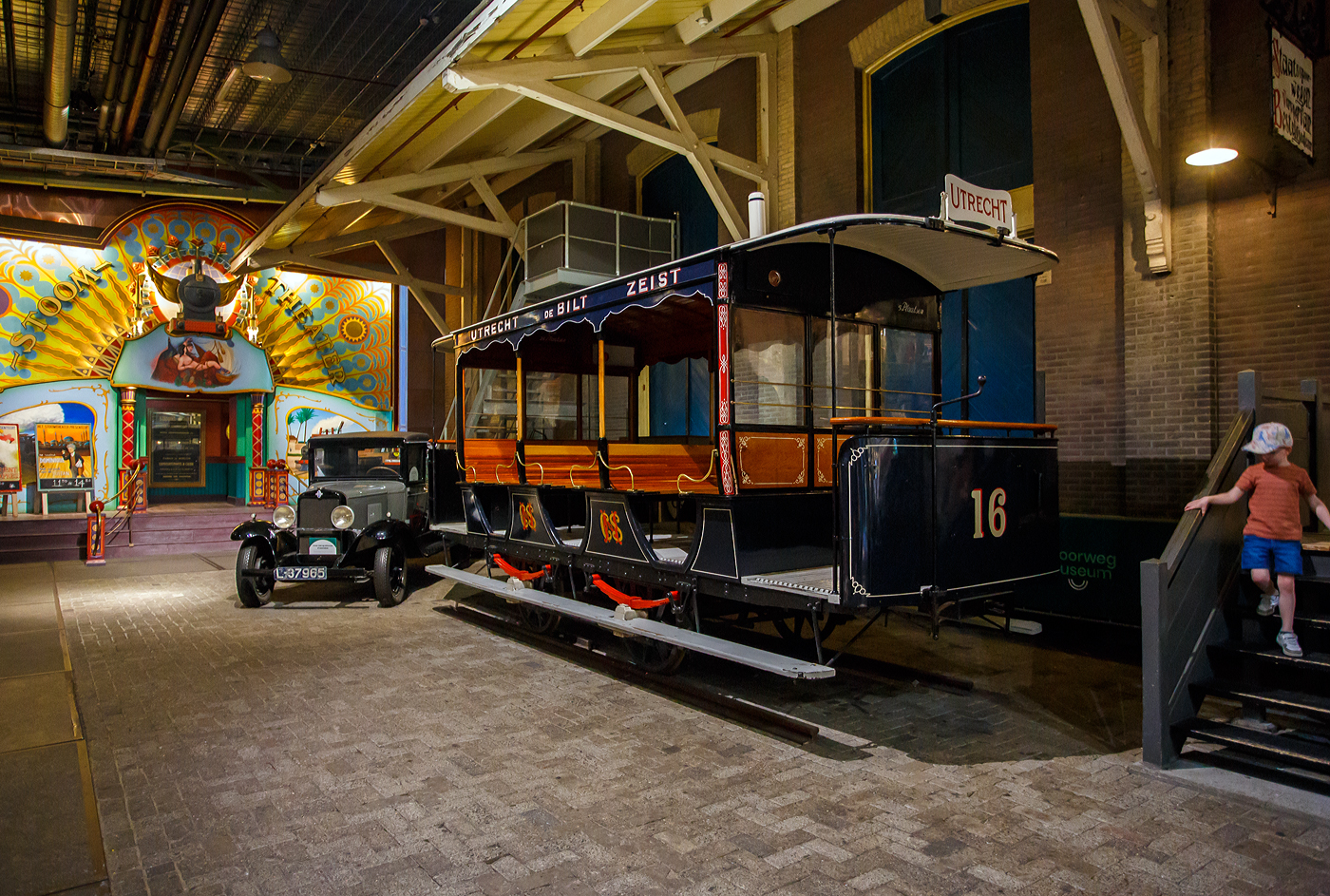 Der zweiachsige Pferdebahnwagen STM 16 der Pferdebahn Utrecht - De Bilt – Zeist der ehemaligen STM - Stichtsche Tramway-Maatschappij, ab 1901 NBM - Nederlandsche Buurtspoorweg-Maatschappij, am 29 April 2025 im Het Spoorwegmuseum (bis 2005 NSM - Nederlands Spoorwegmuseum / Niederländischen Eisenbahnmuseum) in Utrecht - Maliebaan im ehemaligen Bahnhof Maliebaan. Ein Eisenbahnmuseum das wahrhaft zum Verweilen einlädt, leider fehlte uns dieses Mal die Zeit dafür.

Der Wagen wurde 1891 von Beijnes, offiziell Koninklijke Fabriek van Rijtuigen en Spoorwagens J.J. Beijnes (Königliche Fabrik für Kutschen und Eisenbahnwagen J.J. Beijnes) in Haarlem gebaut und an die damalige STM - Stichtsche Tramway-Maatschappij geliefert.

Ende des 19. und Anfang des 20. Jahrhunderts verkehrten in der Stadt und Provinz Utrecht bereits Straßenbahnen der ersten Generation. So u.a. von 1879 bis 1949 eine Überlandstraßenbahn Utrecht - De Bilt – Zeist, betrieben zunächst durch die STM Stichtsche Tramway-Maatschappij, übernommen im Jahr 1901 durch die Nederlandsche Buurtspoorweg-Maatschappij. Diese war bis 1909 eine Pferdebahn beförderte Fahrgäste vom Utrechter Hauptbahnhof durch die Voorstraat und Biltstraat in das schicke Städtchen Zeist. Ab 1909 wurde sie eine elektrische Überlandstraßenbahn. So wurde dieser Anhänger ab 1909 meist im Sommer von der elektrischen Straßenbahn gezogen. Die Überlandstraßenbahn war teilweise auch für die Schließung des Bahnhofs Maliebaan, dem heutigen Eisenbahnmuseum, verantwortlich. Viele Fahrgäste aus Richtung Hilversum wählten zum Ein- und Aussteigen die Haltestelle Biltstraat. Von dort aus bot diese Bahn eine bessere Verbindung ins Stadtzentrum von Utrecht, sodass es keinen Grund mehr gab, weiter zum Bahnhof Maliebaan zu fahren. 

Die STM 16 ist das älteste noch erhaltene Beispiel einer wichtigen Phase in der Geschichte des niederländischen Schienenverkehrs, der Überland-Pferdebahnlinien. Der zweiachsige Wagen/Anhänger war bis 1946 in Betrieb.

Quellen: Het Spoorwegmuseum und nmld.nl
Stand/Überarbeitung: September 2025
