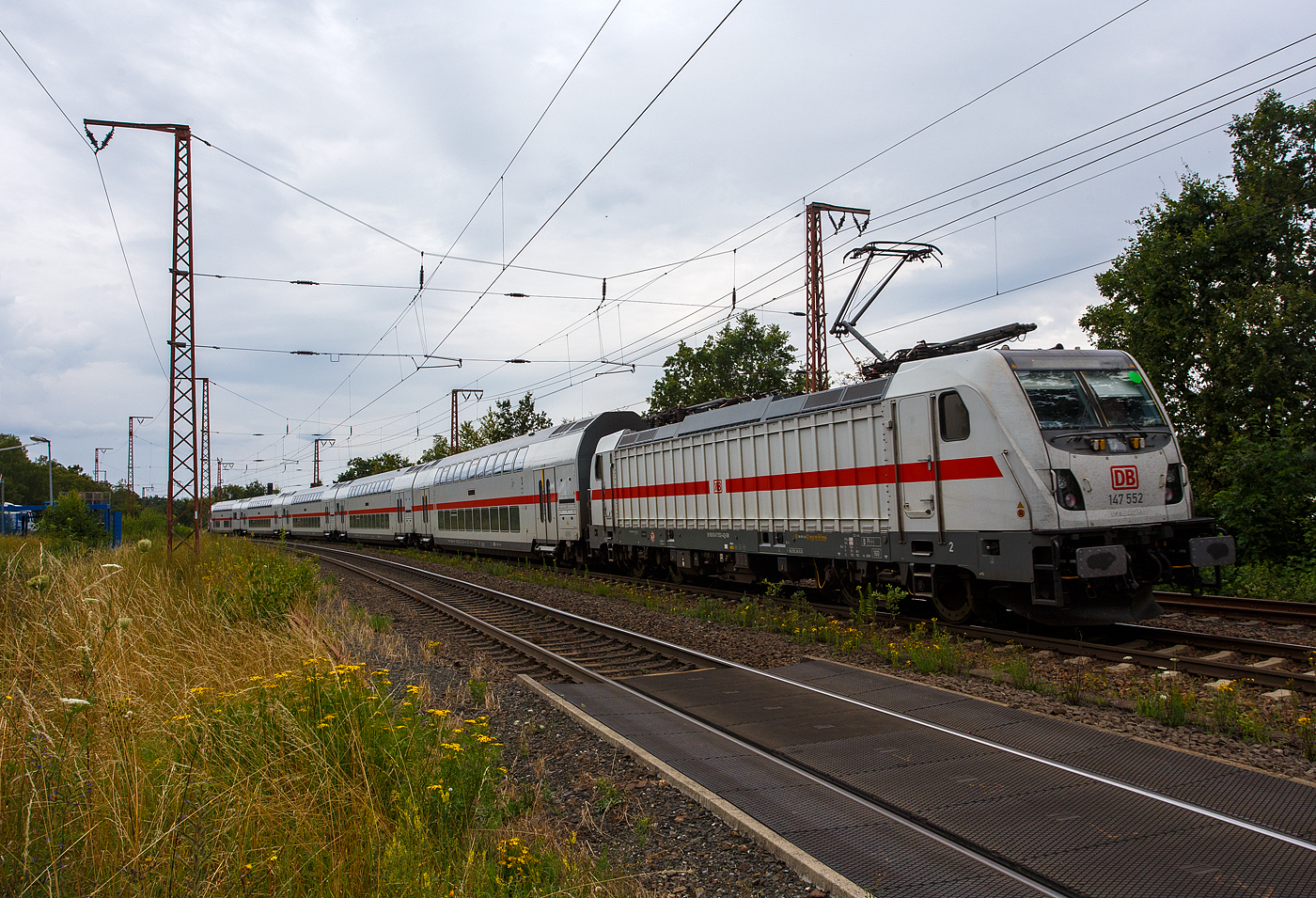 Die 147 552-4 (91 80 6147 552-4 D-DB) der DB Fernverkehr AG schiebt die IC2-Garnitur 4890, als IC 2320 / RE 34 (Frankfurt/Main Hbf – Dillenburg - Siegen - Dortmund Hbf), Steuerwagen voraus am 01 August 2024 durch Rudersdorf (Kr. Siegen) in Richtung Siegen. 

Die TRAXX P160 AC3 wurde 2016 von Bombardier in Kassel unter der Fabriknummer 35224 gebaut und an die DB Fernverkehr AG geliefert. Sie hat die Zulassungen für Deutschland und die Schweiz, daher hat sie auch vier Stromabnehmer. Der Bahnstrom (15.000 V 16 ⅔ Hz) ist ja derselbe, aber die Palettenbreite (Wippe) hat im SBB-Netz eine Breite von 1.450 mm und somit 500 mm schmaler als die Wippen fürs DB Netz (1.900 mm breit). Das Schleifleistenmaterial ist bei beiden aus Graphit.