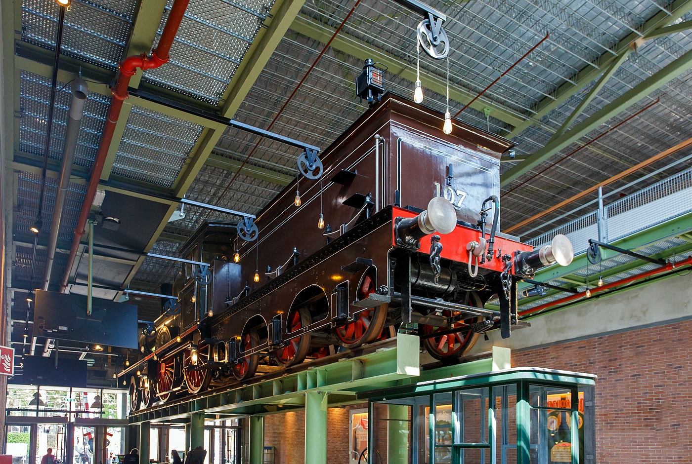 Die 1889 gebaute 2'B Schnellzug-Dampflokomotive NRS 107 „Rhijnboog“ (Rheinbogen), später HSM 353, NS 1604 (NS-Baureihe 1600), am 29 April 2025 hoch aufgeständert im Het Spoorwegmuseum (bis 2005 NSM - Nederlands Spoorwegmuseum / Niederländischen Eisenbahnmuseum) in Utrecht im ehemaligen Bahnhof Maliebaanstation.   

Die Dampflok wurde 1889 von Sharp, Stewart & Co. in Glasgow (GB) unter der Fabriknummer 3563 gebaut und an die NRS - Nederlandsche Rhijnspoorweg-Maatschappij (Niederländische Rheinbahn-Gesellschaft). Im Jahr 1890 wurde die NRS vom niederländischen Staat übernommen, der ihren Betrieb an die Staatsbahnen (SS) übertrug. Das NRS-Rollmaterial wurde zwischen den Staatsbahnen (SS) und der Hollandse IJzeren Spoorweg-Maatschappij (HSM) aufgeteilt, so wurde die NRS 107 zur HSM 353. Ab 1921, nach der Zusammenlegung des Rollmaterials der SS und der NRS, wurde die Lok zur NS 1604.

Die Baureihe NS 1600 war eine Reihe von 59 Schnellzugdampflokomotiven der NS - Nederlandse Spoorwegen (Niederländischen Eisenbahnen) und ihrer Vorgängergesellschaften, der NRS - Nederlandsche Rhijnspoorweg-Maatschappij (Niederländische Rheinbahn-Gesellschaft), der SS - Maatschappij tot Exploitatie van Staatsspoorwegen (Gesellschaft zur Nutzung der Staatseisenbahnen) und der HSM - Hollandsche IJzeren Spoorweg-Maatschappij (Niederländischen Eisenbahn-Gesellschaft). Die Loks wurden von Sharp, Stewart & Co. in Glasgow (GB) gebaut.

Die Loks vom Typ „Rhijnboog“ stellt einen großen Sprung nach vorn im Lokbau dar. Es war die erste Lokomotive, die vorne ein führendes Lauflachsen-Drehgestell hatte, das die Fahrleistung verbesserte. Dies ermöglichte auch die Länge und damit die Kapazität des Kessels zu erhöhen. Sie gehörten damals zu den größten Lokomotiven Europas.

Der Spitzname  Rhijnboog  kombiniert den Namen der niederländischen Eisenbahngesellschaft, die die Lokomotive (Nederlandsche Rhijnspoorweg-Maatschappij) mit dem Wort  Bogie  bestellt hat. Nach der Auflösung der NRS wurde die Lokserie schließlich von der HSM übernommen. Sie waren mit diesen Loks so zufrieden, dass sie 50 weitere nahezu identische Lokomotiven bestellte. Die Lokomotive NRS 107 hat einen besonderen Platz im Museum, sie überragt die Besucher von seinem erhabenen  Thron . 

NRS 101 bis 109:
Im Jahr 1889 bestellte die Nederlandsche Rhijnspoorweg-Maatschappij (NRS) neun Lokomotiven mit den Nummern 101–109 bei der Fabrik von Sharp Stewart and Company in Glasgow. Die Achsfolge der Lokomotiven war 2'B. Zum Zeitpunkt der Bestellung waren dies die ersten niederländischen Lokomotiven mit einem Laufdrehgestell vor den Antriebsrädern (Triebachsen). Sie waren die letzten von der NRS bestellten Lokomotiven. Ein Jahr später, 1890, wurde die NRS aufgelöst und die Ausrüstung zwischen der Niederländischen Eisenbahn-Gesellschaft (HSM) und der Staatlichen Eisenbahn-Ausbeutungsgesellschaft (SS) aufgeteilt. Die Lokomotiven 101, 102, 103, 108 und 109 gingen an die SS und erhielten die SS-Nummern 1101–1103 und 1108–1109. Die 104-107 gingen an das HSM und bekamen die HSM-Nummern 350-353. Da die Lokomotiven für die kleinen Drehscheiben der SS zu lang waren, übergab die SS die fünf Lokomotiven ein Jahr später, 1891, an die HSM, wo sie die Nummern 354–358 erhielten. Aufgrund des Drehgestells für die Antriebsräder, auch Bogie genannt, und der Herkunft des Rhijnspoor erhielten diese Maschinen bei der HSM den Spitznamen „Rhijnboog“.

HSM 359 bis 408:
Die HSM war mit den von ex NRS Lokomotiven so zufrieden, dass sie 40 weitere bei Sharp Stewart and Company bestellte. Diese wurden zwischen 1891 und 1892 unter den Nummern 359–398 in Dienst gestellt. Im Jahr 1900 wurde eine weitere Bestellung über 5 Einheiten mit den Nummern 399-403 bei Sharp Stewart and Company aufgegeben. Schließlich wurde im Jahr 1903 die Klasse 404–408 in Betrieb genommen, die von der North British Locomotive Company (Atlas Works) gebaut wurden, da Sharp Stewart & Co. Company inzwischen zu dieser Gesellschaft fusioniert war.

Bedingt durch den Ersten Weltkrieg wurde 1917 eine Zweckgemeinschaft der unabhängigen Eisenbahngesellschaften HSM (Hollandsche IJzeren Spoorweg-Maatschappij) und SS (Maatschappij tot Exploitatie van Staatsspoorwegen) gegründet. Die beiden Gesellschaften errichteten einen gemeinsamen Bahnbetrieb, blieben aber rechtlich unabhängig. Dies änderte sich 1938, als HIJSM und SS vollständig fusionierten und in der AG NS (Nederlandse Spoorwegen, früher Nederlandsche Spoorwegen geschrieben) aufgingen. Das Rollmaterial wurde bereits 1921 zusammengelegt, so wurden die Lok zur NS 1604 nummeriert.

Die NS 1614, 1633, 1636, 1637 und 1646 wurden 1921 mit einem Überhitzer ausgestattet. Es war beabsichtigt, die gesamte Serie damit auszurüsten, aber aufgrund enttäuschender Ergebnisse blieb es bei diesen fünf Lokomotiven. Sie wurden in den Jahren 1936–1949 außer Dienst gestellt. Die Lokomotive NS 1622 wurde als letzte dieser Baureihe im Jahr 1949 ausgemustert. Die NS 1604, ex HSM 353, ex NRS 107 „Rhijnboog“ blieb bis 1940 im Einsatz blieb. Die 1604 wurde zur Erhaltung für das Niederländische Eisenbahnmuseum in Utrecht ausgewählt und im Lokschuppen in Maastricht eingelagert, wo die Lokomotive am 8. September 1944 durch Kriegseinwirkung schwer beschädigt wurde. Nach dem Zweiten Weltkrieg wurde die Lokomotive von der NS-Hauptwerkstatt in Tilburg vollständig restauriert und in die braune Version der NRS mit der ursprünglichen Nummer 107 zurückversetzt. Seit März 1955 ist die NRS 107 im Eisenbahnmuseum ausgestellt. Nach der Sanierung des Eisenbahnmuseums in den Jahren 2003 bis 2005 wurde die Lokomotive aufgeständert.	

TECHNISCHE DATEN:
NRS-Serie: Rhijnboog, ab 1921 NS 1600
Hersteller: Sharp, Stewart & Co. in Glasgow (GB), später Atlas Works
Gebaute Anzahl: 59
Spurweite: 1.435 mm (Normalspur)
Achsfolge: 	2'B (engl. 4-4-0), gekuppelt mit dreiachsigem Tender
Länge über Puffer: 16.585 mm 
Treibraddurchmesser: 2.016 mm (neu) 
Laufraddurchmesser (Lok und Tender): 1.105 mm (neu) 
Breite: 2.222 mm
Höhe: 4.267 mm
Höchstgeschwindigkeit: 90 km/h
Rostfläche: 2,15 m²
Gesamtheizfläche: 111,6  m²
Kesselüberdruck: 10,3 kg/cm²
Anzahl der Zylinder: 2 (Ø 457 mm x 660 mm Hub)
Steuerung: Stephenson
Eigengewicht: 81,3 t (Lok 48,8 t und Tender 32,5 t) 
Anhängelast: 4.950 kg
Wasservorrat: 10,2 m³ 
Kohlevorrat: 2,8 t
