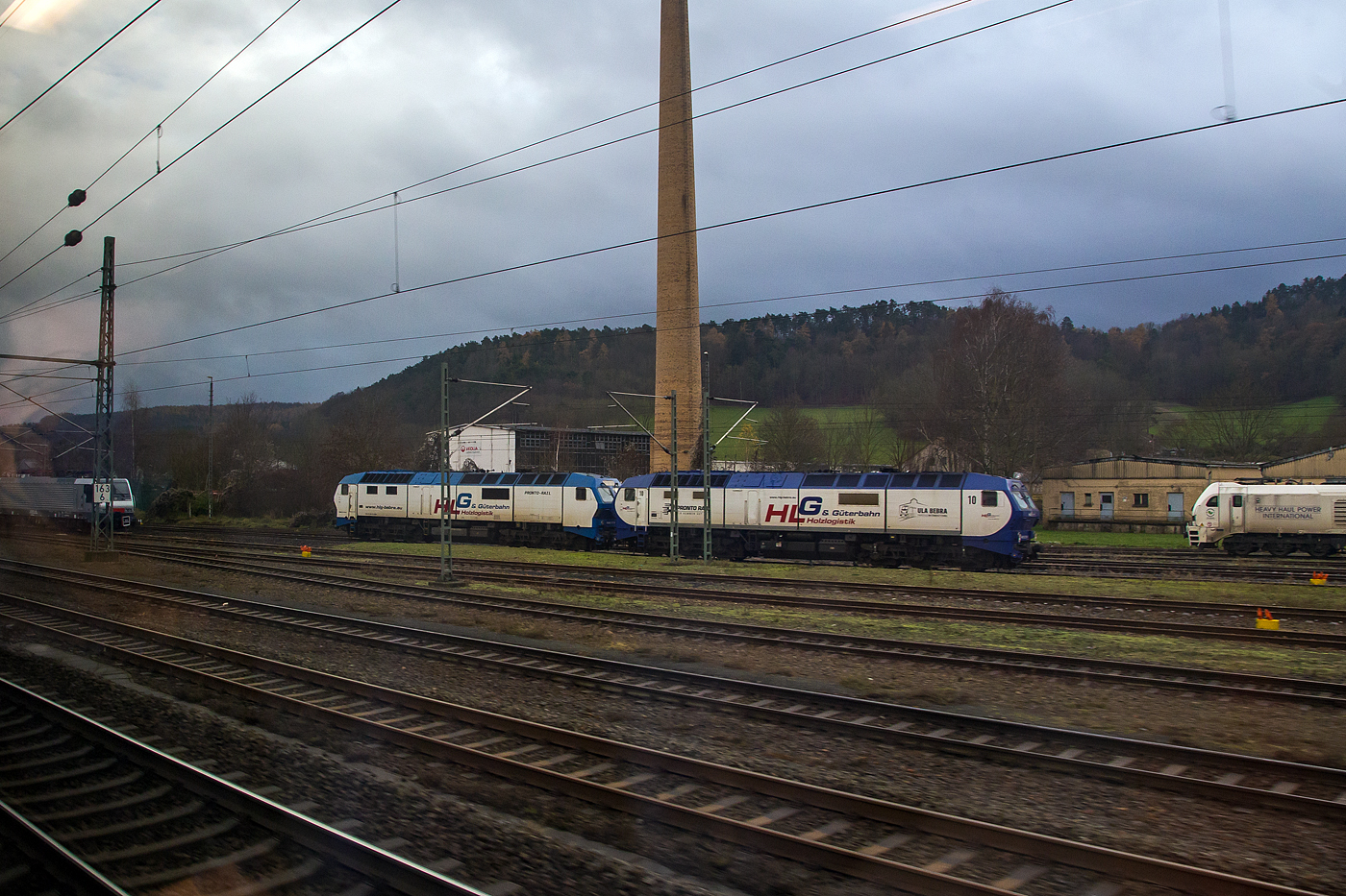 Die DE 2700-01 (92 80 1251 001-4D-BEBRA) und DE 2700-10 (92 80 1251 010-5 D-BEBRA) beide der HGL - Holzlogistik und G�terbahn GmbH, Bebra / Pronto Rail stehen am 08 Dezember 2020 bei der Werkstatt der Uwe Adam EVU GmbH in Eisennach (Aufnahme auch einem ICE). 

Die Loks wurden 1996 von der SFT - Siemens Schienenfahrzeugtechnik GmbH / MaK - Maschinenbau Kiel in Kiel, die DE 2700-01 unter der Fabriknummer 30005 und die -10 unter der Fabriknummer 30014. Im Fr�hjahr 1997 erfolgte die Auslieferung an die NSB BA Oslo NSB Di 6.661 bzw. 670. Die Di 6 war eine Diesellokomotiv-Baureihe der Norges Statsbaner. Sp�ter wurde sie unter verschiedenen Bezeichnungen in Deutschland eingesetzt.

Geschichte:
Konzeptionell stammt die DE 2700 von der MaK DE 1024 (DB-Baureihe 240) ab, erhielt aber durch den Eigent�merwechsel Leistungselektronik der Firma Siemens.Urspr�nglich waren die Loks f�r den Personenverkehr auf der Nordlandsbane Trondheim–Bod�  der NSB vorgesehen. Sie erf�llten die in sie gesetzten Erwartungen im Betriebsdienst jedoch nicht. Vor allem der klimatisch anspruchsvolle Winter in Norwegen machte ihnen zu schaffen. Unter anderem gerieten Lokomotiven in Brand und die Steuerungselektronik funktionierte nicht. Da Nachbesserungen zu keinem Erfolg f�hrten, gab die NSB Anfang 1999 die aus ihrer Sicht unbrauchbaren Dieselloks an den Hersteller Siemens zur�ck.

Nach einer �berarbeitung bei der DSB in Kopenhagen wurden die Lokomotiven im damals Siemens eigenen Mietpool Dispolok eingestellt. Die Loks liefen hier als ME 26. Die luxemburgische Staatsbahn CFL mietete sechs der Loks an, die sie als Ersatz f�r die Elektrolokomotiven der Baureihe 3600 dringend ben�tigten.

Mitte 2004 liefen die Mietvertr�ge aus, und die Fahrzeuge wurden an Vossloh verkauft und nun in DE 2700 umbenannt. Vor der Ablieferung an den neuen Einsatzort bei der Nord-Ostsee-Bahn (NOB) wurden die Loks gr�ndlich aufgearbeitet und modernisiert. Es wurde eine Wendezugsteuerung eingebaut.

Die Loks wurden zun�chst alle bei der NOB f�r Reisez�ge von Hamburg nach Westerland auf Sylt eingesetzt. Hier machten sie in den ersten Monaten durch zahlreiche Ausf�lle sowie einen Generatorbrand im Bahnhof Klanxb�ll von sich reden. Diese Probleme lie�en sich jedoch beheben. Im Laufe des Jahres 2007 wurden verbesserte Schalld�mpfer eingebaut, da das „Wummern“ des knapp 150 Liter gro�en V-12-Motors zu Beschwerden von Anwohnern der Strecke gef�hrt hatte.

Die Fahrzeuge der HLG wurden mittlerweile auf LED-Beleuchtung umger�stet, sodass die unteren Leuchten wegen umstellbarer Farben nur noch einfach vorhanden sind.

Dieselpower der alten Schule
Drei dieser alten Damen (DE2700-01, DE2700-06 & DE2700-10) befinden sich derzeit in der Flotte der HLG. Die DE2700-10 wurde Ende 2021 umfangreich durch die Werkstatt Uwe Adam in Eisenach �berholt und erhielt in diesem Zuge eine neue Lackierung. Die Lokomotiven werden regelm��ig im n�heren Umkreis von Bebra, auf verschiedenen Strecken wie z.B. Immelborn, Wartha, Emleben oder im Kalirevier an der Werra, eingesetzt. Der Sound, den der V12 - Motor mit knapp 150l Hubraum und �ber 3.600 PS generiert, zaubert so manchem Eisenbahner ein L�cheln ins Gesicht.

TECHNISCHE DATEN:
Spurweite: 1.435 mm
Achsfolge:  Co'Co'
L�nge �ber Puffer: 20.960 mm
Drehzapfenabstand: 11.750 mm
Achsabstand im Drehgestell: 3.940 mm
Treibraddurchmesser: 1.060 mm (neu)
H�he: 4.385 mm
Breite: 3.000 mm
Dienstgewicht: 122,2 t
H�chstgeschwindigkeit: 140 km/h
Leistung: 2.650 kW (3.600 PS)
Anfahrzugkraft: 410 kN
Motorbauart: V12-Dieselmotor mit Direkteinspritzung (Anlassen �ber Pressluft)
Motorentyp: MaK 12 M 282
Nenndrehzahl: 1.000 U/min
Tankinhalt : 5.000 l
Antrieb: Dieselelektrisch
Anzahl der elektr. Fahrmotoren: 6
Kleinster befahrbarer Gleisbogen: R 100 m
