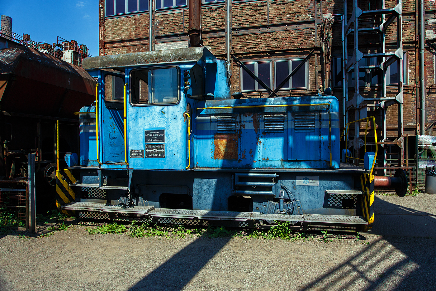 Die Dieselhydraulische Normalspur-Lokomotive Nr. 34, eine Henschel DHG 240 B  Esslinger , der Völklinger Hütte (ehem. Röchling'schen Eisen- und Stahlwerke GmbH in Völklingen/Saar, ab 1982 ARBED-Saarstahl GmbH, ab 1986 Saarstahl Völklingen GmbH) hier am 01 Juli 2025 abgestellt als Denkmal, mit zwei selbstentladende Schüttgutwagen der Gattung Fad oder Falns am Eingang der Rohstoffentladebunker in Richtung Hüttenbahnhof. Die Darstellung steht für den Rohstofftransport der Hütte zur Herstellung des Sintergutes. Die Lok war auch auf Bundesbahn Gleisen zugelassen. Heutiger Eigentümer der Lok und Wagen ist die Initiative Völklinger Hütte e.V..

Die zweiachsige dieselhydraulische Lok mit Antrieb über Gelenkwellen wurde 1969 von Henschel in Kassel unter der Fabriknummer 31210 gebaut und an die Röchling'sche Eisen- und Stahlwerke GmbH in Völklingen als Lok 34 geliefert. Weiterer Lebenslauf/Umfirmierungen: 
01.01.1971 > Stahlwerke Röchling-Burbach GmbH, Völklingen  34  
Juni 1982 	 > ARBED-Saarstahl GmbH, Völklingen  34  
Juni 1986 	 > Saarstahl Völklingen GmbH, Völklingen  34  
10.05.1989 > Saarstahl AG, Völklingen  34  
Später an MGS - Metallurgische Gesellschaft Saar mbH, Völklingen  34  
März 1999 	an Weltkulturerbe Völklinger Hütte - Europäisches Zentrum für Kunst und Industriekultur GmbH, Völklingen und Aufstellung als Denkmal. 

Die Lokomotive war bis zu einem Motorschaden 1997 in verschiedenen Bereichen eingesetzt: Transport von Duplexeisen zwischen der Mischerhalle und dem Siemens-Martin-Werk, im Bereich des Siemens-Martin-Werks, der Elektroöfen und des Hammerwerks sowie nach Einbau einer Funkfernsteuerung im Hafenbereich der Saarstahl AG.

Mit den Fahrzeugen der Typenreihe DHG 160 B / DHG 200 B / DHG 240 B und DHG 350 B bot Henschel Anfang der 1960er Jahre Gelenkwellenmaschinen für den unteren Leistungsbereich an, deren konstruktiver Ursprung nicht in Kassel, sondern in Esslingen der lag. Im Jahr 1961 übernahm Henschel das Diesellok-Typenprogramm der Maschinenfabrik Esslingen. Es wurde eine bei der Maschinenfabrik Esslingen als Typ Bern in Auftrag gegebene Lokomotive als DHG 325 in Kassel fertig gebaut und an den Auftraggeber, die Duisburger Kupferhütte, ausgeliefert. Der Typ Bern stellte auch die Basis für das oben genannte Typenprogramm dar. Dieses unterschied sich zwar optisch deutlich von seinem Esslinger Ursprung, übernahm aber die wesentlichen konstruktiven Merkmale der Fahrzeuge aus Esslinger Produktion. Gleichzeitig gab es keinen technischen und optischen Bezug zu den bisher im eigenen Henschel-Typenprogramm angebotenen Fahrzeugen.

Bei den neuen Gelekwellenfahrzeugen gab es leistungsmäßig deutliche Überschneidungen mit dem kleinsten Typ der 3. Generation, der stangengetriebenen DH 240 B. Diese wurde dann in der 4. Generation durch die DHG 240 B ersetzt.

Die Henschel DHG 240 B wurde zwischen 1963 und 1971 in 34 Exemplaren gebaut. Die Loks gingen vornehmlich an Chemiebetriebe und Raffinerien in Deutschland und der Schweiz. Von diesen wurden elf Loks explosionsgeschützt ausgerüstet. Einzelne Loks wurden ebenfalls mit Funkfernsteuerung ausgerüstet. Mit weitem Abstand am stückzahlenstärksten ist die DHG 240 B, die im Gegensatz zu ihren schwächeren  Schwestern  nicht mit einem DIWABUS-Getriebe von Henschel, sondern mit einem Voith-Hydraulikgetriebe ausgestattet ist. Zahlreiche Fahrzeuge stehen auch heute noch im täglichen Einsatz.

TECHNISCHE DATEN:
Baujahre: 1963 bis 1971
Gebaute Stückzahl: 34
Spurweite:	1.435 mm (Normalspur)
Achsfolge: B
Länge über Puffer: 7.160 mm
Achsabstand: 2.800 mm
größte Breite: 3.000 mm
größte Höhe über Schienenoberkante: 3.600 mm
Raddurchmesser: 850 mm (neu)
kleinster befahrbarer Gleisbogen: 35 m
Eigengewicht: 28 t
Max. Achslast: 16 t
Dieselmotor: wassergekühlter 6 Zylinder / 4-Takt-Diesel-Reihenmotor mit Turboaufladung, ohne Ladeluftkühlung vom Typ Henschel 6R 1416 A, Hubraum 13,3 l.
Leistung: 177 kW (240 PS)
Drehzahl: 1.600 U/min
Höchstgeschwindigkeit: 30 / 60 km/h
Leistungsübertragung: hydraulisch (Voit-Getriebe)
Kraftstoffvorrat: 400 l
