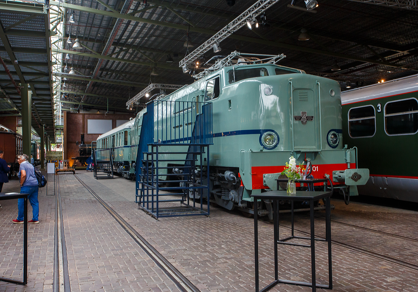 Die Elektrolokomotive NS 1201 der NS-Baureihe 1200 am 24 Juni 2025 im Het Spoorwegmuseum (bis 2005 NSM - Nederlands Spoorwegmuseum / Niederländischen Eisenbahnmuseum) in Utrecht im ehemaligen Bahnhof Maliebaan. Ein Eisenbahnmuseum das zum Verweilen einlädt.

Die Lok wurde 1951 von Werkspoor in Utrecht unter der Fabriknummer 928 gebaut (Baldwin-Lizenz) und an die NS - Nederlandse Spoorwegen (Niederländischen Staatsbahnen, heute Niederländische Eisenbahnen AG) geliefert. Wobei die Entwicklung und Konstruktion, sowie auch einige Teile, von Baldwin Locomotive Works in Philadelphia (USA) stammt. Auch die Entwicklung der Elektrotechnik stammt aus den Vereinigten Staaten und zwar von Westinghouse Electric Corporation (Monroeville), diese wurde von Heemaf in Hengelo (Westinghouse-Lizenz) geliefert.

Die Baureihe 1200 der Niederländischen Eisenbahnen (NS) ist eine sechsachsige Elektrolokomotivbaureihe aus den Jahren 1951 bis 1953. Die Lokomotiven waren bis 1998 bei der NS im Einsatz.

Die Nase war der erste Teil dieser unglaublich starken und robusten Lokomotive, der ins Auge fiel. Sie sollte dem Lokführer bei Kollisionen einen besseren Schutz bieten. Diese Lokomotiven wurden in den Niederlanden nach einem amerikanischen Design und mit einigen amerikanischen Teilen gebaut, die teilweise durch amerikanische Dollar über den Marshall-Plan finanziert wurden. Die NS nutzte diese Lokomotiven, um sowohl Personenzüge als auch Güterzüge zu ziehen. Ihre wichtigste Aufgabe war es Schnellzüge zu ziehen. Als sich die NS 1995 trennte, wurden alle 1200er Jahre an NS Cargo übertragen und begannen im Güterverkehr zu arbeiten. 

Nach Ende des Zweiten Weltkrieges gab es großen Fahrzeugmangel bei der NS. Da man keine Erfahrungen mit dem Bau elektrischer Lokomotiven hatte, kaufte man Lokomotiven bei verschiedenen Lieferanten. Die bei Baldwin konstruierten Lokomotiven wurden bei Werkspoor in Utrecht gefertigt. Die bei Westinghouse entwickelte Elektrotechnik wurde von Heemaf in Hengelo geliefert. Von den ursprünglich 75 georderten Lokomotiven wurden nur 25 ausgeliefert. Der amerikanische Einfluss ist an den hochliegenden Führerständen und den großen, dem Schutz des Lokführers dienenden Vorbauten erkennbar. Am 30. November 1950 wurden 25 Loks bestellt, am 27. September 1951 konnte die erste Probefahrt stattfinden. Am 9. Februar 1952 wurde die 1201 offiziell in Dienst gestellt, der Preis pro Lok betrug 765.000 NFL (ca. 347.000 Euro).

Die Loks hatten sechs Motoren des Typs TM94. Die ersten 14 Lokomotiven waren türkis lackiert, danach setzte sich wegen des sichtbaren Abriebs der Stromabnehmer eine rotbraune Farbgebung durch. Ab 1954 erhielten alle Loks eine blaue Farbgebung, 1971 erhielt die erste Lok die neuen NS-Hausfarben grau/gelb, 1983 war auch die letzte Lok entsprechend umlackiert. Am 30. März 1998 endete der planmäßige Einsatz bei der NS.

Fünf weitere Lokomotiven (1214, 1215, 1218, 1221, 1225) wurden 1999 an die ACTS Nederland BV verkauft. Diese vermietete 2010 drei Lokomotiven (1214, 1215, 1225) an die Eisenbahngesellschaft Euro-Express Treincharter (EETC). Von der wurden sie bis April 2015 unter anderem vor den Nachtzügen Niederlande–Österreich zwischen Amsterdam und Venlo eingesetzt. Fünf Maschinen (1201, 1202, 1211, 1218, 1221) sind als Museumslokomotiven erhalten. NS 1211 ist Botschafterlok im Bahnpark Augsburg. Leihgeber ist das Eisenbahnmuseum Utrecht.

TECHNISCHE DATEN:
Hersteller mech.: Werkspoor (in Lizenz von Baldwin)
Hersteller Elektrik: Heemaf (in Lizenz von Westinghouse)
Baujahre: 1950 bis 1953
Ausmusterung bei NS: bis 1998 
Gebaute Anzahl: 25
Nummerierung: NS 1201 bis 1225
Spurweite: 1.435 mm (Normalspur)
Achsfolge: Co’Co’ 
Länge über Puffer: 18.086 mm
Höhe: 4.064 mm
Treibraddurchmesser: 1.100 mm (neu)
Dienstgewicht: 108 t
Antriebsmotoren: 6 Stück Heemaf vom Typ TM 94
Getriebeübersetzung: 20 : 71
Höchstgeschwindigkeit: 130 km/h (Planmäßig) / 150 km/h (technisch möglich)
Stundenleistung: 6 x 392 kW = 2.350 kW
Dauerleistung: 6 x 367 kW = 2.200 kW
Anfahrzugkraft: 195 kN
Dauerzugkraft: 164 kN bei 80 km/h / 64,7 kN bei 123 km/h
Stromsystem: 1,5 kV DC (=)
Kleister befahrbarer Gleisbogen: R 100 m
