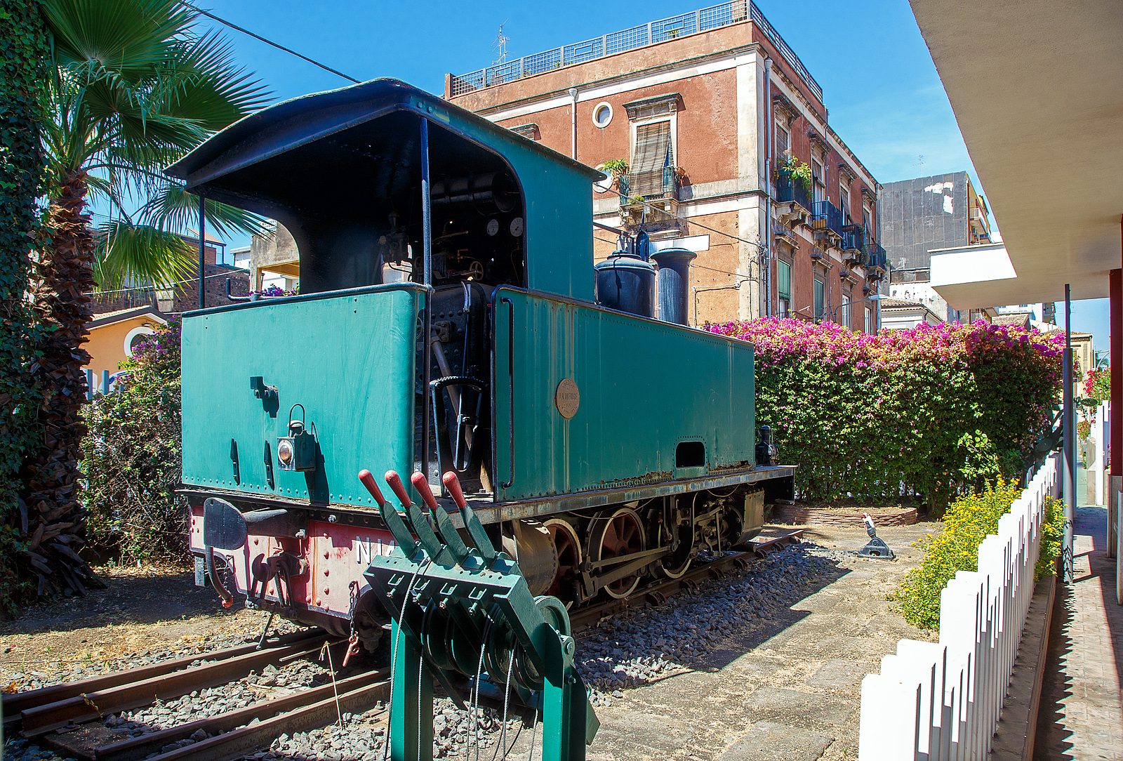 Die F.C.E. N. 14 „LA MEUSE“ der Ferrovia Circumetnea (FCE), ex V 2 der Ferrovia Alifana, steht äußerlich aufgearbeitet, neben einem Wasserkran auf einer Weiche aus der Gründerzeit, neben dem Empfangsgebäude des Bahnhofes Catania-Borgo, hier am 17.07.2022.

Die dreiachsige kohlebefeuerten Schmalspur-Dampflok wurde 1909 von der Société Anonyme des Ateliers de Construction de la Meuse in Liége (Lüttich) unter der Fabriknummer 2224 gebaut und als V 2 an die Ferrovia Alifana in Piedimonte Matese, für ihre 950 mm - Schmalspurbahn von Piedimonte Matese nach Napoli, heute Teil der EAV - Ente Autonomo Volturno S.r.l. (Linie 16). Die Serie bestand aus drei Dampflokomotiven der Gruppo (Gruppe) V 1-3. Die Lok 2 wurde dann 1926 an die Ferrovia Circumetnea (FCE) in Catania verkauft. Bei der Circumetnea sind noch zwei Dampflokomotiven vorhanden, die Nr. 10 (ex. MASCALI) und hier im Bild zu sehende Nr. 14 „LA MEUSE“, ex V 2 der Ferrovia Alifana.

Die 3 belgischen Lokomotiven wurden als Gruppe V 1 bis 3 eingereiht. Sie zeichneten sich durch sehr kleine Räder mit einem Durchmesser von 758 mm (neu) aus. Im Gegensatz zu Circumetnea gab die Ferrovia Alifana ihnen jedoch keinen Namen. Die Lokomotive Nr. 14 ist die einzige der drei, die überlebt hat, und ist als Denkmal im Bahnhof Catania-Borgo ausgestellt. Die anderen beiden Lokomotiven, die Mitte der 1950er Jahre stillgelegt wurden, wurden einige Jahre später abgebrochen.

Die Lokomotiven waren einfach aufgebaute C-Kuppler-Tenderloks (0-3-0 T), mit außenliegenden 2-Zylindern mit Heusinger-Steuerung (oder Walschaerts-Steuerung). Sie waren mit einer Westinghouse-Druckluftbremse und einer Handfeststellbremse ausgestattet.

TECHNISCHE DATEN:
Hersteller: La Meuse in Liége (Lüttich)
Gebaute: 3
Spurweite: 950 mm
Achsfolge: C
Länge: 6.070 mm
Achsabstand: 2 x 1.900 mm = 3.800 mm
Treibraddurchmesser: 758 mm (neu)
Eigengewicht: 17 t
Dienstgewicht: 22 t