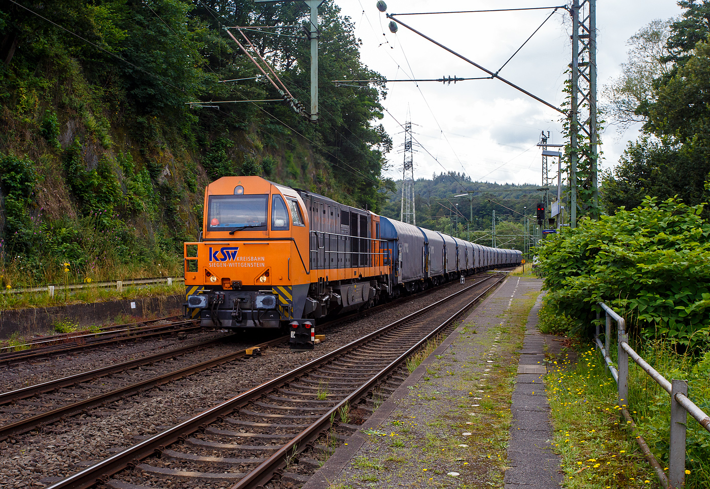 Die KSW 43 (92 80 1273 018-2 D-KSW), die asymmetrische Vossloh G 2000 BB der KSW (Kreisbahn Siegen-Wittgenstein) fährt am 01 Juli 2024, mit einem beladen Coilzug (16 Stück VTG Coilwagen der Gattung Shimmns-ttu, VTG Bez. F46.520D) vom Hafen Duisburg kommend, durch den Bahnhof Scheuerfeld(Sieg) in Richtung Herdorf via Betzdorf. 

In Betzdorf (Sieg) geht es dann über KBS 462 „Hellertalbahn“ nach Herdorf zum Rbf der KSW (Betriebsstätte FGE -Freien Grunder Eisenbahn). Für die Weiterfahrt auf den Pfannenberg (in Neunkirchen-Salchendorf) über nimmt dort dann die KSW 47 (92 80 1271 027-5 D-KSW), ex D 2 der HFM, eine Vossloh G 1000 BB, den Zug.