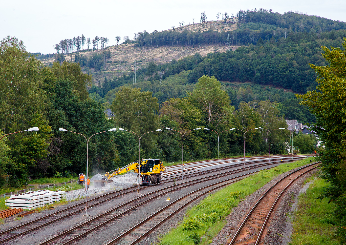 Die KSW (Kreisbahn Siegen-Wittgenstein) lässt, auf ihren Rangierbahnhof in Herdorf der Betriebsstätte FGE -Freien Grunder Eisenbahn (KSW NE447 / DB-Nr. 9275), nach und nach die Gleise erneuern. 
Hier ist am 24.08.2023, der Caterpillar CAT M323F Zweiwegebagger mit hydrostatischem Schienenradantrieb (Serien-Nr. RH600 189), Kleinwagen Nr. D-ZBM 99 80 9902 521-0 der ZEPPELIN Baumaschinen GmbH (Garching), ein Mietbagger vermietet an die Firma  W. Hundhausen Bauunternehmung GmbH (Siegen), mit dem Einschottern eines Gleises beschäftigt.

Rechts die DB Strecke KBS 462 „Hellertalbahn“. 
