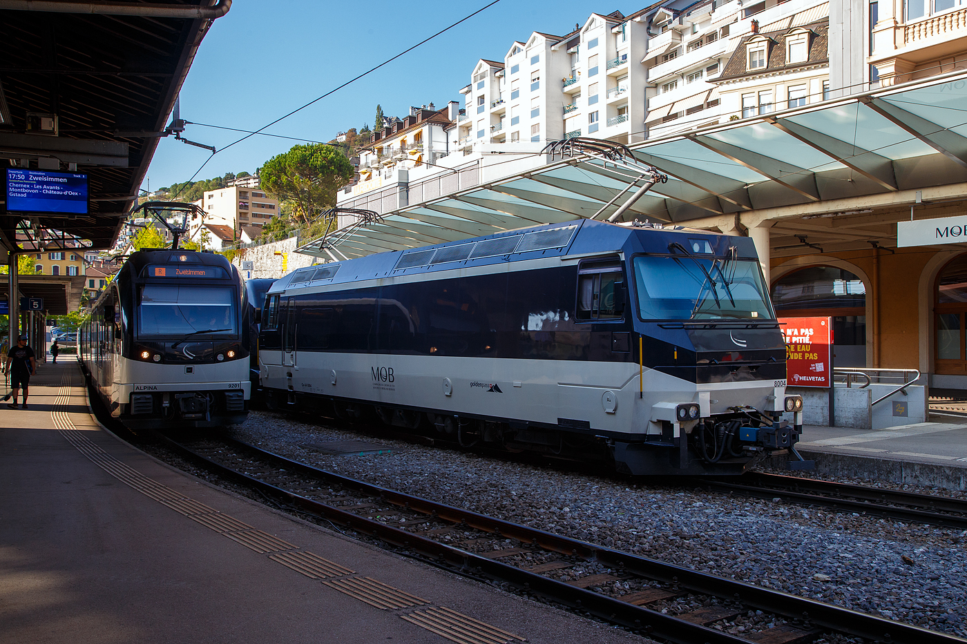 Die MOB Ge 4/4 8004 steht am 10 September 2023 mit einem GPX - GoldenPass Express (Stadler GPX-Wagen mit variablem Drehgestellen) im Bahnhof Montreux.

Die Lok wurde 1994 von der Schweizerische Lokomotiv- und Maschinenfabrik (SLM) in Winterthur gebaut, die elektrische Ausrüstung ist von ABB Verkehrssysteme AG. Die Ge 4/4 sind die leistungsstärksten schweizerischen Gleichstrom-Triebfahrzeuge. Die Maschinen der MOB (Montreux–Berner Oberland-Bahn) haben übrigens keine Eigennamen oder Wappen, sie tragen nur die Betriebsnummer. Eine der vier Lok, die Lok 8003 wurde 2019 an die Rhätische Bahn verkauft und wurde dort zur RhB Ge 4/4 653 umgebaut.

Die vier 1994 von SLM und ABB für die MOB gebauten Ge 4/4 ähneln sowohl optisch als auch mechanisch den Ge 4/4 der Bière-Apples-Morges-Bahn (BAM) und den Ge 4/4 III der Rhätischen Bahn (RhB). Der elektrische Teil musste jedoch dem Betrieb ab Fahrleitung mit 900 V Gleichspannung angepasst werden und eine allfällige Umrüstung auf Zweisystem-Betrieb mit zusätzlich 15 kV 16,7 Hz berücksichtigen. Der Betrieb mit Wechselspannung wäre notwendig gewesen, wenn die GoldenPass-Linie mit einer dritten Schiene bis Interlaken verlängert worden wäre. So ist im Lokkasten bereits der Platz für einen Transformator für den Betrieb bei 15 kV mit 16 2/3 Hz Wechselstrom vorgesehen, ebenso lässt sich ein dritter Pantograph nachrüsten.

Die Lok ist mit vier Schnellschaltern ausgerüstet, die über Drosseln die wassergekühlten Stromrichter versorgen. Die Eingangsspannung der Wechselrichter kann von 630 bis 1080 Volt variieren, für den Zweispannungsbetrieb hätte die Zwischenkreisspannung 1350 Volt betragen. Dies erforderte eine Anpassung der Fahrmotorwicklung gegenüber den RhB-Loks, die für eine Zwischenkreisspannung von 2100 bis 2800 Volt ausgelegt sind. Der mechanische Teil des Fahrmotors ist baugleich mit demjenigen der RhB-Motoren.

Die Loks haben eine Leistung von 2000 kW und erreichen eine Höchstgeschwindigkeit von 120 km/h, die jedoch im Normalbetrieb mangels geeigneter Strecken nicht erreicht wird. Wegen der relativ geringen Spannung sind sehr hohe Stromstärken erforderlich. Daher fahren die Loks der Montreux–Berner Oberland-Bahn (MOB) immer mit zwei Stromabnehmern am Fahrdraht. Zudem sind die Stromabnehmer mit drei statt den üblichen zwei Schleifleisten versehen, was man hier im Bild auch gut sehen kann.

Möglich ist auch die Vielfachsteuerung mit den MOB GDe 4/4, was erst erforderlich ist, wenn mehr als sechs Panoramawagen zu befördern sind. Dies ist bisher nur vereinzelt vorgekommen.

Die Loks hatten (wie im Bild auch zu sehen) Mittelpuffer mit einer Schraubenkupplung, ab 2017 wurden die drei bei der MOB verbliebenen Loks auf automatische Mittelpufferkupplung (Schwab-Kupplung) umgebaut. Seit 2020 sind die verbliebenen 3 Lokomotiven mit allen Fahrzeugen Steuerwagen sowie den Triebwagen mit Schwab-Automatikkupplung in Vielfachsteuerung kompatibel und

TECHNISCHE DATEN:
Gebaute Anzahl: 4, die Ge 4/4 8003 wurde 2019 an die RhB verkauft.
Hersteller mech. Teil: SLM, Winterthur
Elektr. Ausrüstung: ABB Verkehrssysteme AG
Spurweite: 1.000 mm (Schmalspur)
Achsfolge: Bo'Bo'
Länge über Puffer: 16.100 mm
Treibraddurchmesser: 1.070 mm (neu)
Breite max.: 2.800 mm
Höhe : 3.860 mm
Dienstgewicht: 63 t
Anzahl der Fahrmotoren: 4
Maximale Leistung am Rad: 2.400 kW
Dauerleistung am Rad: 2.000 kW
Dauerzugkraft am Rad: 150 kN
Anfahrzugkraft am Rad: 200 kN
Höchstgeschwindigkeit: 120 km/h
Anhängelast bei 45 km/h und 73‰ Steigung: 170 t
Anhängelast bei 80 km/h und 25‰ Steigung: 300 t
Stromsystem: 900 V DC (Gleichstrom)
Stromübertragung: 2 Einholmstromabnehmer mit je 3 Schleifleisten
