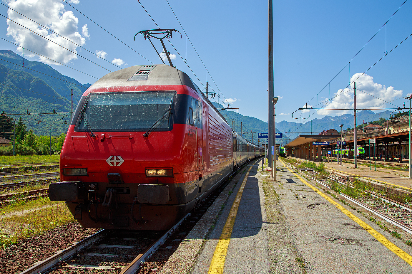 Die SBB Re 460 016-0  Rohrdorferberg Reusstal   (91 85 4 460 016-0 CH-SBB) hat am 26 Mai 2023 mit dem IR 3017 (Brig – Domodossola) den Zielbahnhof Domodossola (I) erreicht und ist nun mit dem Zug auf Gleis 6 abgestellt. Nach gut 2 ½ Stunden (um 13:48 Uhr) geht es dann als IR3022 von Domodossola zurück nach Brig.

Die Lok2000 wurde 1992 von der SLM (Schweizerischen Lokomotiv- und Maschinenfabrik in Winterthur) unter der Fabriknummer 5477 gebaut, der elektrische Teil ist von der ABB (Asea Brown Boveri). Sie war 2015 der 2. Prototyp der im SBB Werk Yverdon-les-Bains (VD) das vollständige Modernisierungs-/Refit-Programm LOK2000 durchlief. So sollen die Loks fit für die nächsten 20 Jahre sein, zudem werden durch alle 119 SBB Re 460 Lokomotiven so etwa 30 Gigawattstunden Energie eingespart.

Modernisierungs-/Refit-Programm der LOK2000 (Re 460):
Zwischen 2015 und November 2022 unterzog die SBB alle ihrer 119 Re 460 Lokomotiven (LOK2000) einem Refit-Programm (Modernisierung) im SBB Werk Yverdon-les-Bains (VD). Dabei wurden unter anderem die auf dem GTO-(Thyristor)-Halbleiter-Technologiestand der 1990er-Jahre basierende Leistungselektronik (GTO- Traktionsumrichter) durch IGBT-Traktionsumrichter (insulated-gate bipolar transistor) der neuesten Generation von ABB ersetzt, welche die Fahrmotoren mit Strom speisen und Bremsenergie zurückgewinnen. Die SBB hat damit, in Zusammenarbeit mit der ABB, die gesamte Flotte Re 460 modernisiert und hat diese auf den neuesten Stand der Antriebstechnik gebracht. Die gesamte Flotte spart dank neuen Traktionsumrichtern und weiteren technischen Optimierungen jedes Jahr rund 30 Gigawattstunden Energie ein, das sind fünf Gigawattstunden mehr als ursprünglich erwartet. Die Energieeinsparungen entsprechen dem durchschnittlichen Stromverbrauch von 10.000 Schweizer Haushalten. Die Umrüstung auf modernste IGBT-Technologie und mit Wasser statt Öl betriebene Kühlsysteme der Umrichter ermöglicht eine Senkung des Energieverbrauchs und sorgt für einen ökologischeren und sicheren Bahnbetrieb.

Geschichte und Beschreibung:
Die SBB bestellte im Zusammenhang mit den Projekten Bahn 2000 eine erste Serie von 12 Universal-Hochgeschwindigkeitslokomotiven, noch als Re 4/4VI – 10701 bis 10712 bestellt. Diese Bestellung wurde noch vor Beginn der Konstruktionsarbeiten auf 24 erhöht. Für den alpenquerenden Güterverkehr wurde von der Eidgenossenschaft eine weitere Serie von 75 Lokomotiven (auch als 'Hupac'-Lokomotiven bekannt) in Auftrag gegeben. Die vorgegebenen Liefertermine für dieses Los waren derart kurz, dass die SLM an die Kapazitätsgrenze ihrer Werkstätte gelangte. Es wurden deshalb dreißig Lokkasten an Krauss-Maffei in München untervergeben 

Im Hinblick auf die geplante Hochgeschwindigkeitsstrecke wurden nochmals zwanzig Lokomotiven nachbestellt, was die Serie auf 119 ansteigen ließ. Die im Volksmund Lok 2000 genannte Lokomotive hat eine Höchstgeschwindigkeit von 230 km/h und eine Leistung von 6,1 MW und ist für den Schnellzugs- wie Güterverkehr ausgelegt. Um die Gesamtmasse von 84 Tonnen nicht zu überschreiten musste ein gesickter Leichtbaukasten konstruiert werden, dessen Form vom Designstudio Pininfarina entworfen wurde.

Um auf den Strecken durch die Alpen den Radverschleiß gering zu halten, haben die Lokomotive radial einstellbare Achsen erhalten. Auch auf dem Gebiet der elektrischen Ausrüstung wurde Neuland beschritten. Zwei Ebenen Steuerelektronik für das Fahrzeug und den Antrieb sind eingebaut. Über das Fahrzeugleitgerät wurden zu viele Elemente gesteuert, wie Scheibenwischer oder Signalhorn. Dies führte anfänglich zu vielen Störungen und oft blieben Lokomotiven auf der Strecke liegen. Erst die Verbesserung der Programmierung erlaubte es die Zuverlässigkeit so zu erhöhen, dass es kaum mehr zu Ausfällen führte. Als Feststellbremse wurden in den Drehgestellen Permanentmagnetschienenbremsen eingebaut. Auch hier gab es am Anfang übermäßig viele Störungen und die Lokomotiven klebten regelmäßig an den Schienen fest.

Um den Schiebedienst auf den Gebirgsstrecken weniger personalaufwendig durchzuführen, wurden mehrere Lokomotiven mit einer amerikanischen Funkfernsteuerung ausgerüstet und als Ref 460 bezeichnet. Damit konnte von der Zugspitze aus eine am Zugschluss eingesetzte Lokomotive ferngesteuert werden. Die Divisionalisierung der SBB erlaubte es nicht mehr groß davon zu profitieren, denn alle Lokomotiven wurden dem Personenverkehr zugeteilt und der Einsatz vor Güterzügen war somit beendet.

Die Re 460 blieb nicht ein Einzelstück und entwickelte sich zu einer Lokomotivfamilie. Eine erste Bestellung wurde von der BLS für acht Lokomotiven gemacht. Die BLS Re 465 hat im Gegensatz zur Re 460 vier statt zwei Stromrichter. Jeder Motor hat dadurch seine eigene Regelung um die Eigenschaften im Bergeinsatz zu verbessern. Weiter können die BLS-Lokomotiven mit allen gängigen Schweizer Lokomotiven in Vielfachsteuerung verkehren. Das erlaubte es bei der BLS den Lokomotiveinsatz flexibler zu gestalten. Die SBB bestellte ebenfalls zehn Re 465, die bei Ablieferung schon das blaue BLS-Design hatten, aber erst später von der BLS übernommen wurden. Nach Finnland konnten auch in zwei Serien Lok 2000 verkauft werden. Dort sind sie als Sr2 bezeichnet. Nach Versuchsfahrten in Norwegen im schwierigen Winterdienst bestellten die NSB die fast baugleichen Lokomotive EL18 2241 bis 2262. Eine letzte Lieferung umfasst zwei Lokomotiven für Hongkong. Sie werden an jedem Ende eines Doppelstockzuges eingesetzt. Dies sind die letzten von SLM in Winterthur gebauten Fahrzeuge. Die Re 460 ist bis auf weiteres die letzte Vollbahn-Streckenlokomotive, die komplett von schweizerischen Unternehmen konstruiert und gebaut wurde. Der Lokomotivbau in der Schweiz wurde inzwischen aus wirtschaftlichen Gründen eingestellt. Einzig Stadler Rail stellt in der Schweiz noch Lokomotiven für den Rangier- und für den Zahnradbetrieb her.

TECHNISCHE DATEN der Re 460:
Nummerierung:  Re 460 000–118
Anzahl: 119
Baujahre: 1991–1996
Hersteller: SLM - Winterthur / ABB (Asea Brown Boveri)
Design: Pininfarina
Hersteller Lokkasten: teilweise (30 Stück) Krauss-Maffei (München)
Spurweite: 1.435 mm (Normalspur)
Achsformel: Bo'Bo'
Länge über Puffer: 18.500 mm
Höhe: 4.300 mm
Breite: 3.000 mm
Drehzapfenabstand: 11.000 mm
Achsabstand im Drehgestell:  2.800 mm
Laufraddurchmesser: 1.110 mm (neu)
Dienstgewicht: 84,0 t
Höchstgeschwindigkeit:  200 km/h (zugelassen)  / 230 km/h (Techn. möglich) 
Stundenleistung: 6.100 kW / 8.300 PS
Dauerleistung: 4.800 kW / 6.000 PS
Anfahrzugkraft: 300 kN
Dauerzugkraft: 275kN
Stromsystem: 15 kV, 16,7 Hz (über Oberleitung)