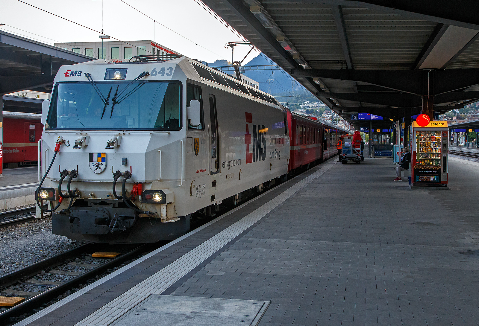 Die wei�e RhB Ge 4/4 III (�BoBo 3�) – 643  Vals  mit Werbung f�r die Ems-Chemie steht am 06.09.2021im Bahnhof Chur mit dem IR (InterRegio) nach St. Moritz zur Abfahrt bereit. 

Durch das ansteigende Verkehrsaufkommen war die Rh�tische Bahn (RhB) veranlasst Ende der 1980er Jahre ein Konzept f�r eine neue Lokomotivgeneration zu erstellen. Gemeinsam mit SLM und ABB entwickelte die Rh�tische Bahn eine Lok, deren Antrieb auf der Drehstromtechnik mit GTO-Thyristoren basiert. Die neuen Lokomotiven sollten in der Lage sein, Personen- und G�terz�ge auf dem Stammnetz, aber auch Autotransportz�ge durch den neuen Vereinatunnel zu ziehen. Wobei f�r den Einsatz im Vereinatunnel eine sechsachsige Lok die ideale Wahl gewesen w�re, wurde entschieden, vierachsige Fahrzeuge zu bestellen. Da die vierachsigen Loks f�r die F�hrung von Z�gen auf den kurvenreichen Strecken des Stammnetzes, wie beispielsweise der Albulalinie, geeigneter sind.

Neben den modernen GTO-Stromrichtern haben die Lokomotiven neu entwickelte Drehgestelle mit selbst�ndig radial einstellenden Rads�tzen, dadurch wird der Verschlei� zwischen Rad und Schiene vermindert.

1989 bestellte die RhB sechs und 1991 weitere drei Lokn bei der Schweizer Industrie. Die Biere - Apples - Morges Bahn (BAM) schloss sich 1990 dieser Bestellung mit zwei Fahrzeugen f�r die Bef�rderung schwerer Panzerz�ge an. Vier weitere Lokomotiven wurden unter Verwendung �hnlicher mechanischer Komponenten f�r die MOB gebaut. Der elektrische Teil der MOB Lokomotiven hingegen ist eine komplette Neuentwicklung. P�nktlich zur Er�ffnung des Vereina-Tunnels wurden von der RhB Ende 1999 drei weitere Lokomotiven in Betrieb genommen.

Ein modernes und funktionelles Design wurde f�r die Gestaltung von dem Lokomotivkasten gew�hlt. Die Antriebseinheit besteht aus zwei modular aufgebauten, je einem Drehgestell zugeordneten, Zweipunkt-GTO-Umrichtern. Der Antriebsstromrichter eines Drehgestells speist zwei parallel geschaltete sechspolige Drehstrom-Asynchrontraktionsmotoren mit Kurzschlussl�ufer. Der unterflur angeordnete Transformator und die Wechselrichter sind �lgek�hlt. Die Leitelektronik wurde von der SBB Re 460 (Lok 2000) abgeleitet und basiert auf dem MICAS-S2 Leitsystem von ABB (sp�ter Adtranz, jetzt Bombardier Transportation).

F�r die Bedienung des Fahrzeuges wurde von der RhB ein v�llig neues Konzept gew�hlt: die Vorgabe der Zug- und Bremskr�fte erfolgt �ber zwei Side-Sticks. Alle wichtigen Messwerte wie Fahrgeschwindigkeit, Zug-/Bremskraft, Fahrleitungsspannung und Bremsleitungsdruck werden auf zwei Displays angezeigt. Diese dienen auch zur Darstellung von St�rungsmeldungen - konventionelle Messinstrumente sucht man in dem F�hrerraum vergeblich. Dieses Konzept konnte sich im Betriebsalltag nicht bew�hren. Die 1999 gelieferten Lokomotiven Ge 4/4 III 650 - 652 (wie diese hier) wurden daher mit einem konventionellen Fahrerpult mit Handrad ausgestattet.

Die Mehrzwecklokomotiven des Typs Ge 4/4 III werden haupts�chlich im Schnellzugdienst auf der Strecke Chur - St. Moritz sowie zur Bef�rderung der Autoz�ge durch den Vereinatunnel eingesetzt.

Technische Daten der Ge 4/4 III:
Betriebsnummern: 641 – 652
Anzahl Fahrzeuge: 12

Ge 4/4 III 641 - 649
Hersteller – mecha. Teil: SLM
Hersteller - Elektrik: ABB Verkehrssysteme AG

Ge 4/4 III 650 - 652
Hersteller – mecha. Teil der : Adtranz (ex. SLM)
Hersteller - Elektrik: Adtranz (ex. ABB)

Baujahre: 1993 (6 St�ck), 1994 (3 St�ck) und 1999 (3 St�ck)

Spurweite: 1.000 mm
Achsanordnung: Bo'Bo'
H�chstgeschwindigkeit: 100 km/h
L�nge �ber Puffer: 16.000 mm
Achsabstand im Drehgestell: 2.400 mm
Triebraddurchmesser (neu): 1.070 mm
Breite: 2.800 mm
Dienstgewicht: 62,0 t
Anh�ngelast bei 4,5% mit 50 km/h: 210 t
Anh�ngelast bei 3,5% mit 60 km/h: 290 t
Anh�ngelast bei 1,5% mit 78 km/h: 350 t
Fahrleitungsspannung: 11 kV, AC 16,7 Hz
Anzahl Fahrmotoren: 4 St�ck vom Typ 6 FRA 5248)
Max. Leistung am Rad: 3.100 kW
Dauerleistung am Rad: 2.400 kW
Max. Zugkraft am Rad: 200 kN
Dauerzugkraft am Rad: 170 kN
Getriebe�bersetzung: 1 : 6,136