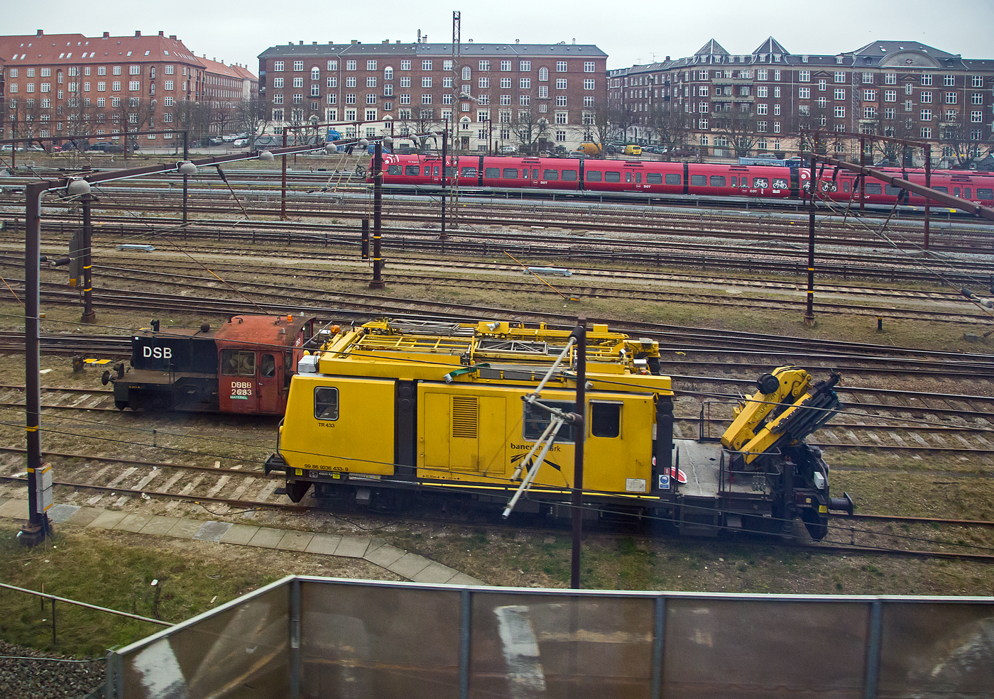 Ein Bild aus dem EC am 20 März 2019 beim Hauptbahnhof Kopenhagen (Københavns Hovedbanegård): Im Vordergrund der Plasser & Theurer Motorturmwagen MTW 10, TR 433 - 99 86 9236 433-9 DK-BDK der Banedanmark (BDK), links dahinter der DSB-Køf-Rangiertraktor Kö ll 263. Im Hintergrund fährt eine S-Bahn bzw. DSB SA 81xx (S-Tog).

Der DSB-Køf-Rangiertraktor:
Der DSB-Køf-Rangiertraktor Kö ll 263 wurde 1966 von der Maschinenfabrik Frichs Maskinfabrik og Kedelsmedie A/S in Aarhus (Århus im Stadtteil Åbyhøj) unter der Fabriknummer 874 gebaut und an die DSB - Danske Statsbaner (Dänische Staatsbahnen) geliefert.

Auf Grund der guten Erfahrungen in Deutschland mit Kleinlokomotiven (Köf) mieteten die Danske Statsbaner im November 1963 die Köf 6645 an, die nach einer Einweisung der DSB-Lokführer, 1964 in Dänemark war. Nach der Erprobung bestellten die DSB 20 Rangiertraktoren, die im Sprachgebrauch in Anlehnung an die deutsche Bezeichnung Køf genannt wurden. Die 251–270 wurden bei Frichs Maskinfabrik og Kedelsmedie A/S in Aarhus (Århus- Åbyhøj) bestellt und 1966 geliefert. Im Gegensatz zur deutschen Baureihe haben die dänischen Køf einen englischen Leyland-Motor. 1967 bestellte die DSB weitere 20 Køf bei Frichs, die 271–290. Die Serie erhielt ein geschlossenes Führerhaus mit Fenstern und Türen und wurde 1968 und 1969 geliefert.

Aufbau:
Das Führerhaus ist auf beiden Seiten mit einem Bedienplatz ausgestattet und mit der niedrigen Fußbodenhöhe von etwa 400 mm über Schienenoberkante dem Ein- und Aussteigen beim Rangieren angepasst. Der Sechszylinder-Viertakt-Motor von Leyland leistet 128 PS (94 kW) bei 1800/min. Sein Kühlwassersystem besitzt eine Vorwärmeinrichtung mit 2.000 W bei 110 V. Der Motor ist über eine Kardanwelle mit dem Voith-Hydraulikgetriebe L33u verbunden. Vom Voith-Getriebe geht die Kraftübertragung über ein Wendegetriebe von Gmeinder und Rollenketten e auf beide Achsen.

Vom Motor, der über ein Handrad bedient wird, werden über Riemen Kompressor und Generator angetrieben. Das Bremsventil für den Zug liegt auf der rechten Führerstandsseite, die Lok allein kann über ein links und rechts vorhandenes Bremsventil gebremst werden.

Die ersten 20 Lokomotiven wurden mit offenen Führerhäusern ohne Türen und Seitenfenster geliefert. Die zweite Serie wurde mit einem vollständig geschlossenen Führerhaus geliefert, ab 1968 erfolgte die Nachrüstung der ersten Serie.

Ursprünglich waren alle Fahrzeuge an beiden Enden mit einer automatischen Rangierkupplung ausgerüstet, die beim Anfahren an einen Wagen automatisch kuppelte und vom Führerstand durch Druckluft entkuppelt werden konnte. Für Zugfahrten wurde sie hochgeklappt und die Schraubenkupplung des ersten Wagens mit dem Kupplungshaken der Lok verbunden. Einzelne Lokomotiven erhielten später normale Schraubenkupplungen.

Ursprünglich waren alle Køfs grün, einige erhielten später das rot/schwarze DSB-Farbschema. Bei einigen Maschinen wurde die Betriebsnummer in übergroßen Ziffern zusätzlich auf dem Dach des Führerhauses als sogenannte  Flyvernummer  (Fliegernummer) angebracht, um sie von den Stellwerken aus leichter identifizieren zu können. Ab 1988 verbesserten eine geräuschdämmende Auskleidung des Führerhauses sowie eine kühlwasserbetriebene Heizung die Arbeitsverhältnisse auf den Loks.

Der Einsatz der Køfs erfolgte landesweit im Rangierbetrieb, vereinzelt übernahmen sie auch leichte Güterzüge im Streckendienst. Mit der Übernahme der Gütersparte  DSB Gods  durch  Railion DK  (heute DB Cargo Scandinavia) gelangten 2001 insgesamt 26 Køfs zum neuen Betreiber, 13 verblieben bei der DSB. Die Railion-Køfs wurden unverändert weiter betrieben und lediglich durch aufgeklebte Schriftzüge gekennzeichnet. Aus dem Überbestand endeten einige Köfs als Ersatzteilspender, andere wurden von verschiedenen Privatbahnen und Gleisbauunternehmen erworben, 5 weitere Køfs blieben museal erhalten. Die veräußerten Køfs wurden bei den neuen Betreibern in Stand gesetzt und erstrahlten in einem bunten Strauß neuer Farbschemata.

Zur Verlängerung der Lebensdauer setzte die DSB 2020 in der Werkstatt Fredericia Køf 253, 263, 273 und 290 instand (die zusätzlich beabsichtigte Moderisierung von Køf 270 wurde nicht ausgeführt). Motor, Getriebe und Bremsanlage wurden überholt, die elektrischen Leitungen wurden erneuert und alle Luftkomponenten erhielten eine Revision bei der  Westfälische Lokomotiv-Fabrik Reuschling  in Hattingen. Auch die Scheinwerfer wurden erneuert, wobei Leuchten mit integrierten roten LED-Ringen aus den zeitgleich verschrotteten MR-Triebzügen verwendet wurden. Bei der Neulackierung mit schwarzem Rahmen und grünen Aufbauten wählte das Werkstattteam einen vergleichsweise hellen Grünton, angelehnt an das  British Racing Green . Die Innenräume der Führerstände blieben ohne Renovierung. Insgesamt überschritt das Projekt die bewilligten Ressourcen an Arbeitszeit und Budget deutlich. Weitere Køf-Instandsetzungen wurden ausgeschlossen, da die Werkstatt in Fredericia geschlossen wurde und das beteiligte Personal in Rente ging.

Anfang 2024 kündigte DB Cargo Scandinavia an, ebenfalls 5 ihrer Køfs instandsetzen zu wollen.

TECHNISCHE DATEN:
Anzahl: 40
Hersteller: Frichs in Aarhus
Baujahre: 1966, 1968-1969
Spurweite:  1.435 mm (Normalspur)
Achsfolge:  B
Länge über Puffer: 6.400 mm
Höhe: 2.935 mm
Breite: 3.070 mm
Achsstand : 2.500 mm
Treibraddurchmesser: 850 mm (neu)
Motor: Leyland UE 680, 6-Zylinder –Viertakt-Dieselmotor
Leistung: 94 kW (128 PS) bei 1.800 U/min
Getriebe: Voith L33u
Kraftübertragung: Dieselhydraulisch, d.h. vom Motor über Gelenkwelle auf Voith-Getriebe über ein Wendegetriebe von Gmeinder über Rollenketten auf beide Achsen.
Höchstgeschwindigkeit: 45 km/h
Dienstgewicht: 17,0 t
Tankinhalt: 200 l
