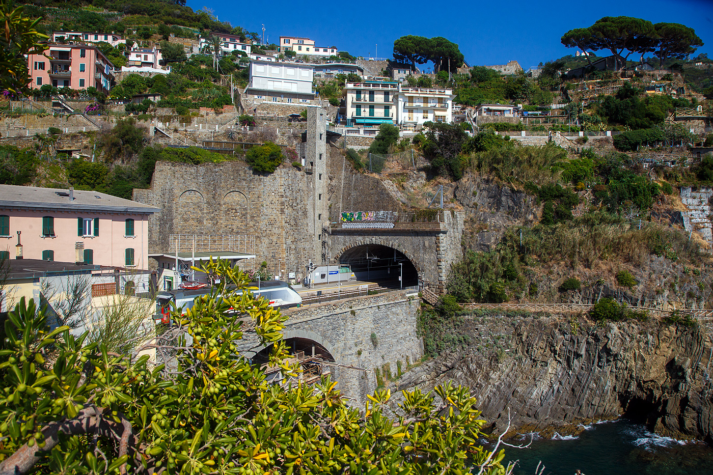 Ein Trenitalia Intercity (IC) rauscht im Sandwich zweier E.414 (ehemalige Triebköpfe E.404 A der ersten ETR 500 – Monotensione) durch den Cinque Terre Bahnhof Riomaggiore in Richtung Genua (Genova).

Hinter dem Zugende auf der anderen Bahnsteigseite sieht am den Eingang von dem Fußgänger-Tunnel vom Bahnhof zum Zentrum von Riomaggiore. Dieser führt durch den Eisenbahntunnel, ist aber abgegrenzt. 

Zudem sieht man hinten auch den hohen Lift (Aufzug), mit diesem kommt man auf den etliche Meter oben liegenden Weg. Der Lift ist aber in der Saison kostenpflichtig, aber er erspart etliche Treppenstufen oder einen gewaltigen Umweg.