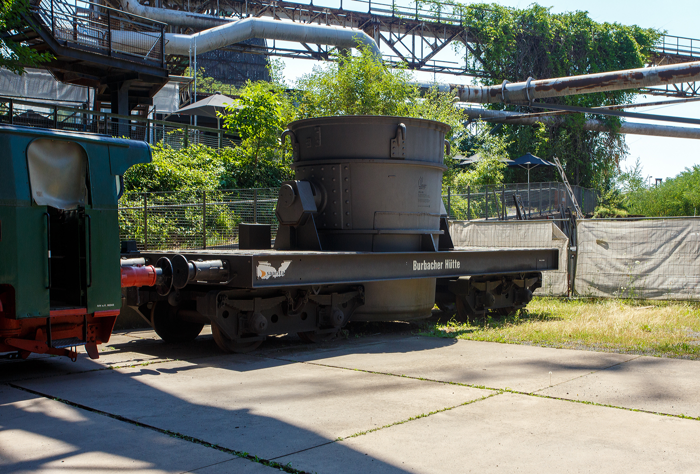 Ein vierachsiger normalspuriger offener Roheisenpfannenwagen der Burbacher H�tte hinter der Normalspur-Dampflokomotive 46, einer Jung Hochofenlok niedriger Bauart, der V�lklinger H�tte (ehem. R�chling'schen Eisen- und Stahlwerke GmbH in V�lklingen/Saar, ab 1982 ARBED-Saarstahl GmbH, ab 1986 Saarstahl V�lklingen GmbH), ausgestellt als Denkmal am Gel�nde V�lklinger H�tte, hier am 01 Juli 2025 am Gel�nde V�lklinger H�tte als Denkmal. Heutiger Eigent�mer ist die Initiative V�lklinger H�tte e.V..

1971 fusionierten die Burbacher und V�lklinger H�tte, und ab 1975 wurde die Stahlproduktion in der Burbacher H�tte eingestellt. Das Bild zeigt einen offenen Pfannenwagen, mit dem das fl�ssige und hei�e Roheisen aus dem Hochofen aufgefangen wird, und dann zur Weiterverarbeitung mittels Werkseisenbahn zur Weiterverarbeitung in das angrenzende Stahl- und Walzwerk weitertransportiert wurde. Da die Entfernungen gering waren, ben�tigte man keine mit Schamott ausgemauerten Torpedopfannenwagen. Ob der Wagen hier auf der V�lklinger H�tte ist mit nicht klar, aber da er erheblich h�her ist als die Lok, so vermute ich das er hier keinen Einsatz fand.