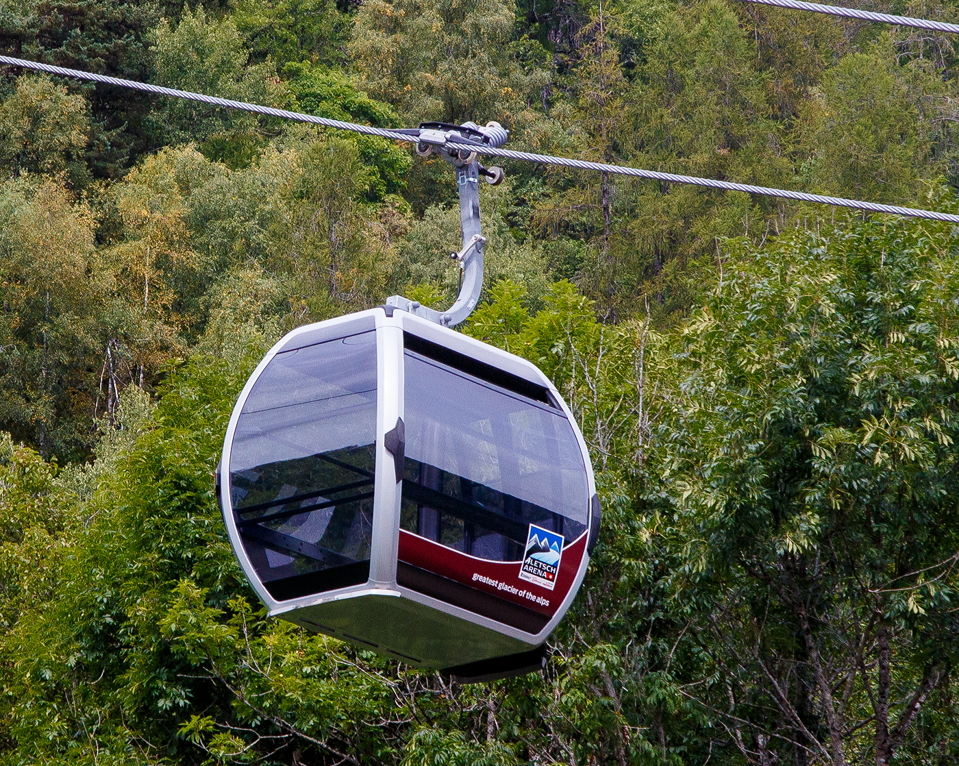 Eine Doppelmayr/Garaventa CWA Omega V Kabinen der neuen 10er-Kabinenbahn Fiesch - Fiescheralp / Kühboden am 07 September 2021in Fiesch aus dem Zug heraus fotografiert.

Hier erkennt man auch oben gut die D-Line Kuppelklemme. Sie vereint die Vorteile der Torsionsfederklemme (DT-Klemme) mit denen der Schraubenfederklemme (A-Klemme) in einer einzigen eleganten Lösung. Diese können bis zu einem Seildurchmesser von 64 mm, sowie 100 % Bahnneigung bei 1.800 kg Fahrzeuggewicht, verwendet werde. Diese kuppelbare Klemmen verbinden das Fahrzeug (Gondel) mit dem Seil. In den Stationen werden sie geöffnet, um das Fahrzeug vom Seil zu lösen, so dass die Fahrgäste bei geringerer Geschwindigkeit ein- und aussteigen können, vor der Ausfahrt aus der Station werden sie dann wieder an das Seil geklemmt.