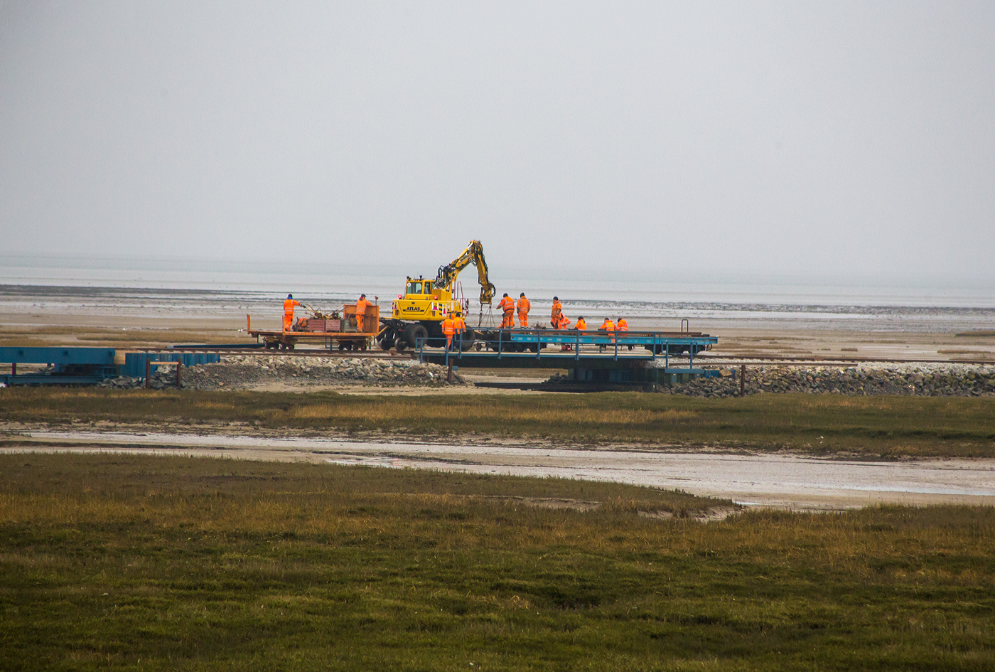 Eine Gleisbau-Rotte ist mit einem Meterspur Zweiwegebagger ATLAS 1404 ZW der Kommanditgesellschaft EMS Gleisbau GmbH & Co. und vorgestellten vierachsigen DB-Flachwagen sowie ein Abrollcontainer auf zwei angehangenen Rollschemeln am Nachmittag des 12 März 2024 zu Gleisbauarbeiten, auf der Wangerooger Inselbahn unterwegs. Hier auf einer Brücke am Steindamm in den Salzwiesen auf halber Strecke zwischen Ort Wangerooge und dem Westanleger.  Das Gleis führt über einen Priel, mehrere Eisenbahnbrücken ermöglichen den Tidefluss (auf- und abfließen Gezeiten) im Priel zwischen der Salinenbucht und der Westlagune.

In den Wintermonaten und jetzt im frühen März sind solche Gleisbauarbeiten im Nationalpark Wattenmeer noch möglich, da noch keine Brutzeit ist. Für die Urlauber besteht aber für die Salzwiesen wie auch für die Dünen ein ganzjähriges Betretungsverbot.

Der ATLAS Zweiwegebagger 1404 ZW-CR für 1.000 mm Spurbreite ausgerüstet, der niedriger Schwerpunkt und quer eingebauter Motor machen ihn standfest auf der schmalen Spur. 

Für den Zweiwege-Schmalspurbagger  ATLAS 1404 ZW-CR  Bagger im Einsatz auf schmaler Spur wurde ein den Anforderungen entsprechendes Fahrgestell gebaut und montiert. Der 1404 ZW ist mit 20 t Dienstgewicht der kleinste ATLAS Zweiwegebagger. Er ist mit einem Deutz Dieselmotor mit Turbolader vom Typ TCD 4.1 (mit 4.038 cm³ Hubraum)  der EU-Stufe V/US-EPA mit 95 KW/130 PS (bei 1.800 U/min) ausgerüstet. Eine Abgasnachbehandlungsanlage mit geschlossenem Diesel-Partikelfilter und kombiniertem SCR-Katalysator sorgen für Umweltfreundlichkeit. Die Linde Load- Sensing-Hydraulik in Verbindung mit dem ATLAS AWE 4 Hydraulikmanagement machen sensibles und millimetergenaues Arbeiten und Fahren möglich.

Das Kontergewicht ist integriert und beim ATLAS 1404 ZW in drei Heckradius- Varianten lieferbar 1.575 mm – 1.700 mm – 1.998 mm. Auf Wangerooge ist die 1.998 mm Variante im Einsatz. Ein niedriger Schwerpunkt und ein quer eingebauter Motor sorgen für optimale Stabilität und Standsicherheit beim Arbeiten auf dem Gleis. Auch beim Heben von wirklich schweren Gewichten bleibt der 1404 ZW trotz Schmalspur sicher und solide. Das Fahrwerk mit robusten, speziell konzipierten Baggerachsen mit Planetengetriebe in allen 4 Radnaben sowie Allradantrieb, Regelfahrmotor mit Lastschaltgetriebe und ein doppelt wirkendes Fahrbremsventil machen sichereres Fahren auf Straße und Schiene möglich. Ein Alleinstellungsmerkmal für ATLAS ist die Starrachse mit einem zuschaltbarem, 100%igem Sperrdifferential. Dieser 1404 ZW wird mit AWE 4 Technik über Reibräder angetrieben. Für diesen Einsatz auf Wangerooge wurden Achsen mit auf 1.000 mm reduzierter Breite montiert.