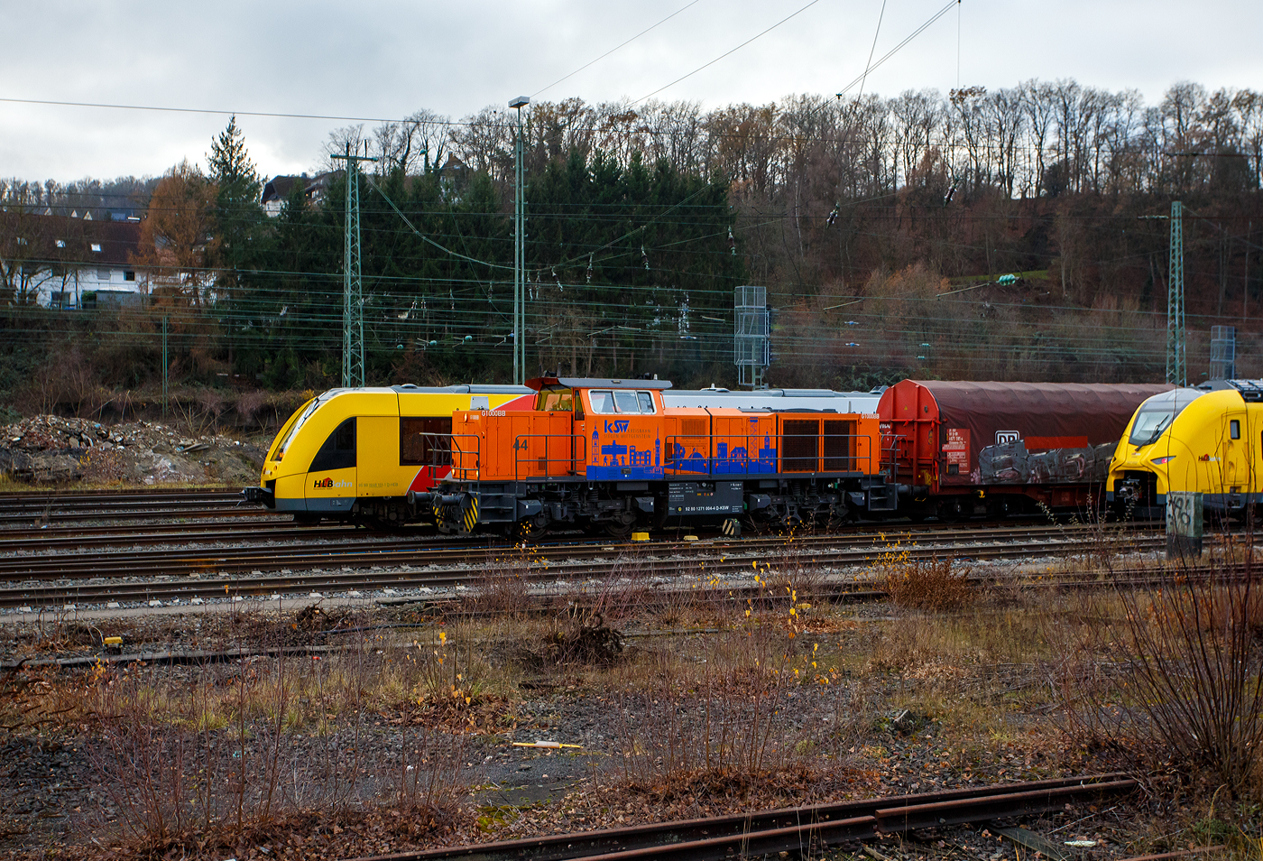 Geführt von der KSW 44 (92 80 1271 004-4 D-KSW), eine MaK G 1000 BB, fährt am 04 Dezember 2025 die KSW (Kreisbahn Siegen-Wittgenstein), im Sandwich mit einem sehr langen und schweren Coilzug, vom Rangierbahnhof Betzdorf/Sieg in Richtung Herdorf zum KSW-Rangierbahnhof (Betriebsstätte FGE - Freien Grunder Eisenbahn) nun los. Hinten (nicht im Bild) ist die nachschiebende KSW 42 bzw. 277 902-3 (92 80 1277 902-3 D-KSW) eine Vossloh MaK G 1700 BB. 

Die Fuhre geht ja über die, nicht elektrifizierte, eingleisige KBS 462 „Hellertalbahn“, da nur eine Güterverkehrsfahrt zwischen den Personenverkehr der RB 96 passt, fährt die KSW lieber eine lange Fuhre im Sandwich. Für eine Lok wäre die Anhängelast eines solchen langen beladenen Coilzuges zu hoch. In Herdorf auf dem Rangierbahnhof der KSW, werden Züge in mehren Teile aufgeteilt und gehen dann über die KSW eigene Strecke NE 447 (ehem. Freien Grunder Eisenbahn nach Neunkirchen-Salchendorf (ehemals Pfannenberger Einigkeit) zu den Produktionshallen der EMW Stahl Service GmbH, ein Geschäftsbereich der SCHÄFER WERKE Gruppe. 

Schon heute werden so dorthin über 300.000 Tonnen Stahl pro Jahr über die Schiene angeliefert, zukünftig sind sogar 600.000 Tonnen geplant.