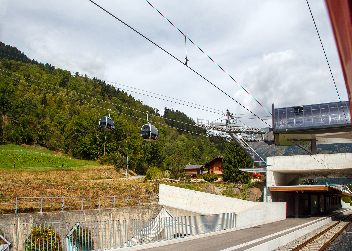 Im MGB Bahnhof Fiesch / neunen ÖV-Hub Fiesch blick am 07 September 2021 aus einem MGB Regio-Zug (R43) heraus. Der Bahnhof ist zugleich Talstation der neuen Doppelmayr/Garaventa-10er-Kabinenbahn der hinauf auf die Fiescheralp.

Links zwei (ja eine berg- und talfahrende) Doppelmayr/Garaventa CWA Omega V Kabinen der neuen 10er-Kabinenbahn Fiesch - Fiescheralp / Kühboden, Baujahr 2019 von der Doppelmayr/Garaventa-Gruppe ist mit 7 m/s Fahrgeschwindigkeit die schnellste 10er-Gondelbahn der Schweiz. Die neue Gondelbahn ersetzt zwei Pendelbahnen von Fiesch zur Fiescheralp, einmal mit 100er-Kabinen und einmal mit 34er-Kabinen. Die Talstation der hochmodernen Anlage befindet sich im neuen ÖV-Hub (Knotenpunkt Öffentlicher Verkehr) in Fiesch, wo direkter Anschluss an die Matterhorn-Gotthard-Bahn und das Postautonetz besteht.
