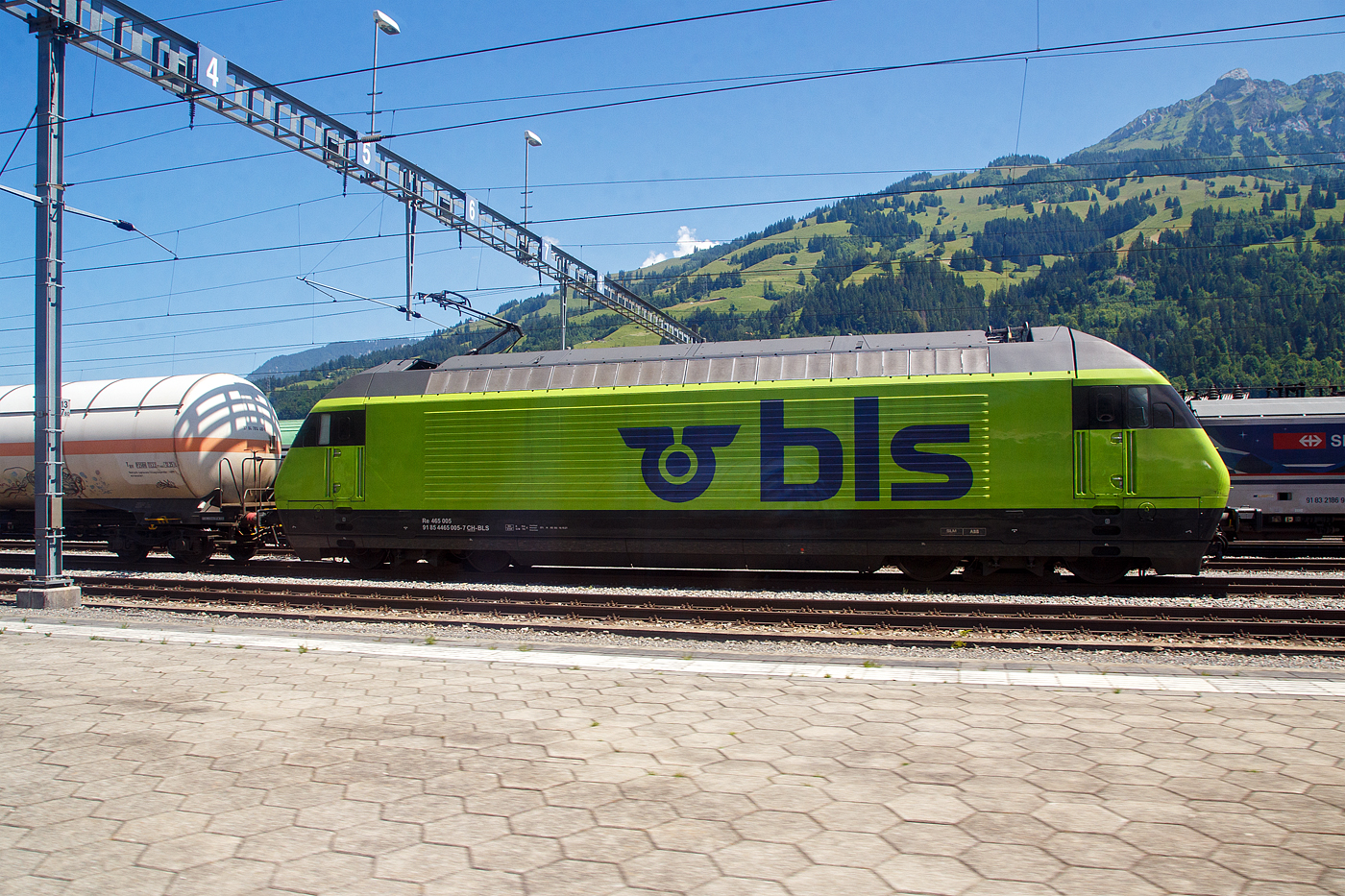 In Frutigen vor dem Lötschberg-Basistunnel wartet am 11 Juli 2022 die BLS Re 465 005, ex „Niesen“ (91 85 4465 005-7 CH-BLS) mit einem Kesselwagenzug auf Weiterfahrt, so konnte ich sie aus einem Zug aufnehmen.

Die Lok wurde 1994 von der SLM (Schweizerische Lokomotiv- und Maschinenfabrik) in Winterthur unter der Fabriknummer 5642 gebaut, der elektrische Teil ist von der ABB (Asea Brown Boveri).

Die Re 465 ist eine von der BLS Lötschbergbahn beschaffte elektrische, vierachsige Universallokomotive mit Umrichtertechnik. Sie ist eine Weiterentwicklung der Re 460 der SBB und gehört zur Lok-2000-Familie. Nach der Erstbestellung der BLS über acht Maschinen die 1994 gebaut wurden, folgten weitere 1997 zehn Loks (009–018) für den Huckepackkorridor. Diese Loks aus den 90er-Jahren sind für den Personen- wie für den Güterverkehr im Einsatz. Künftig werden sie auch beim Autoverlad und im Berner Oberland eingesetzt, nicht mehr im originalen Dunkelblau, sondern im leuchtenden BLS-Grün.

Die Re 465 stellen eine Weiterentwicklung der Re 460 dar. Wie diese hat sie GTO-Thyristoren als Ventile im Stromrichter und Drehstrom-Asynchronmotoren. Der Unterschied liegt in der Umrichteranlage. Im Fall der Re 460 besteht ein Traktionsstromrichter (in der Lok sind zwei Geräte verbaut, ein Gerät pro Drehgestell) aus zwei Vierquadrantenstellern, die den Zwischenkreis parallel versorgen, und einem Motor-Wechselrichter, der zwei Motoren parallel speist. Die Stromrichter der Re 465 gruppieren sich zu drei Vierquadrantenstellern parallel an einem Zwischenkreis mit zwei Wechselrichtern, je einen Motor im Drehgestell speisend. Somit lässt sich eine schleudernde Achse schneller und effektiver wieder in normalen Lauf bringen. Wurde bei der Entwicklung der Re 460 auf Dreipunkt-Stromrichter gesetzt, wurde im Gegensatz dazu die Re 465 mit Zweipunkt-Technik ausgestattet. Die Zwischenkreisspannung senkte man entsprechend von 3,5 kV (Re 460) auf 2,8 kV (Re 465). Zudem besitzt die Re 465 sechspolige Asynchron-Fahrmotoren (Re 460: vierpolige Asynchronmotoren). Daraus resultiert eine gegenüber der Re 460 erhöhte Dauerleistung von 6.270 kW (Stundenleistung von 7.000 kW).

Die Radsätze im Drehgestell stellen sich radial zum Gleisbogen ein.

Zudem war die Re 465 im Unterschied zur Re 460 mit der Vielfachsteuerung des Systems IIId und (mit Übergangskabel) BLS/BBC ausgerüstet. Diese erlaubte die Vielfachsteuerung zum Beispiel mit Re 4/4II, Re 4/4III, Re 6/6, Re 425 oder Ae 415, beim Refit wurde diese Vielfachsteuerung ausgebaut und dafür eine Vielfachsteuerung für TRAXX und Vectron-Loks eingebaut. 

Zwischen 2019 und 2022 durchliefen alle 18 Loks des Typs Re 465 ein Modernisierungsprogramm (Refit). Darin enthalten waren unter anderem der Einbau der Vielfachsteuerung ZMS für den Güterverkehr mit Traxx- und Vectron-Lokomotiven und ETB (Ethernet Train Backbone) für den Autoverlad am Lötschberg. Die Vielfachsteuerung des Systems IIId wurde ausgebaut, so ist die Vielfachsteuerung mit älteren Loks wie der Re 425 nicht mehr möglich. Die Kästen wurden saniert und neu in hellem BLS-Lindengrün mit blauem Logo lackiert. 

Mit diesem Refit verlängert sich die Lebensdauer der rund 25-jährigen Loks um weitere 15 Jahre. Sie sind neu bis ins Jahr 2040 einsetzbar. Die modernisierten Lokomotiven sind gut zu erkennen, sie sind neu grün lackiert werden damit zur auffälligen und klar ersichtlichen BLS-Markenbotschafterin. Das Modernisierungsprogramm kostete insgesamt rund 20 Millionen CHF. Modernisiert wurden alle Loks in der BLS-Werkstätte Bönigen. Im Zuge des Refits wurden alle Taufnamen und Bilder entfernt.

Im Personenverkehr zieht bzw. stößt die Re 465 die GoldenPass-Express-Züge zwischen Interlaken Ost und Zweisimmen, im Güterverkehr wird sie in Mehrfachtraktion mit anderen Loks (TRAXX und Vectron) eingesetzt. Die Loks werden ab Ende 2023 auch beim Autoverlad eingesetzt und ersetzen damit die braunen Loks des Typs Re 425. Damit kann die BLS auf eine Neubeschaffung verzichten.

TECHNISCHE DATEN:
Spurweite: 1.435 mm (Normalspur)
Achsformel: Bo’Bo’
Länge über Puffer: 18.500 mm
Drehzapfenabstand:11.000 mm
Achsstand im Drehgestell: 2.800 mm
Treibraddurchmesser: 1.100 mm (neu)
Höhe: 4.540 mm
Breite: 3.000 mm
Dienstmasse: 84 t
Höchstgeschwindigkeit: 230 km/h
Stundenleistung: 7.000 kW
Dauerleistung: 6.400 kW
Anfahrzugkraft: 300 kN
Stromsystem: Wechselstrom 15 kV 16,7 Hz

Quellen: BLS, Wikipedia und Anschriften
Stand/Überarbeitung: Oktober 2025
