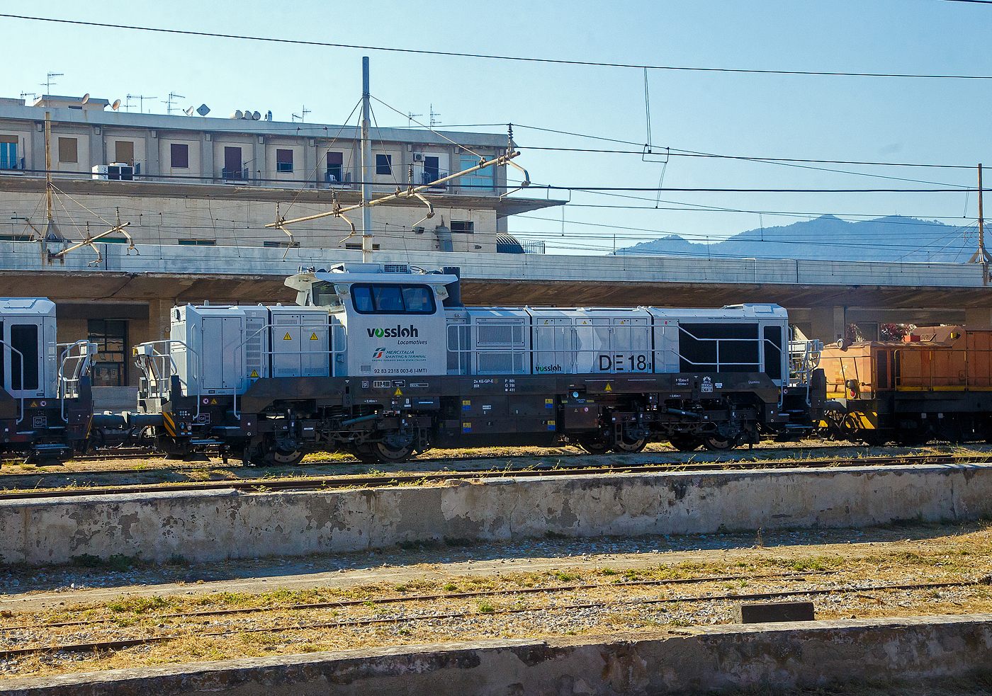 Neue Verschub-Lokomotiven fürs Trajekt-Manöver an den Ufern der Straße von Messina....
Aber im Juli 2022 erledigten diesen Verschub noch die älteren RFI D.145 Loks der Serie 2000.

Links die moderne vierachsige (Bo'Bo') dieselelektrische Vossloh DE 18 2419 „Jasmina“ alias D 318 003-6 (92 83 2318 003-6  I-IMTI) der MIST - Mercitalia Shunting & Terminal S.r.l. am 20.07.2022 abgestellt beim Bahnhof Messina Centrale. Aufnahme aus einem IC Notte.
