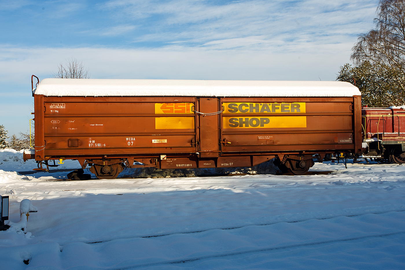 Nicht nur im Süden der Republik hat es etwas geschneit, wie hier auf dem Westerwald auf der Bindweide, aber nicht so mächtig. Der private gedeckte zweiachsige Güterwagen mit Schiebewänden und Schiebedach WEBA 07 - 10 80 0715 001-5 D-WEBA (ex 10 80 071 5 001-5 D-DB) der Gattung Tbis 869 - Bauform A (für Büromöbel der SSI Schaefer Shop GmbH) der Westerwald WEBA abgestellt am 03 Dezember 2023 (am 1. Advent) auf dem Betriebshof der Westerwaldbahn (WEBA) auf der Bindweide bei Steinebach/Sieg. Der Wagen ist auf DB-Strecke Scheuerfeld – Betzdorf(Sieg) zugelassen. 

Beschreibung:
Ab 1966 wurden von der DB bei der Waggonunion Siegen (eigentlich Dreis-Tiefenbach, zuvor Siegener Eisenbahnbedarf A.G.) erste Wagen der Gattung Tbis 869 der Bauform A beschafft. Bei dieser Bauform konnte das Dach nur von einer kleinen oben liegenden Bühne an den Stirnwänden geöffnet werden. Zwei Jahre später wurde die Bauform B dieser Gattung gefertigt. Hierbei war es nun möglich das Dach mittels Handräder an einer Stirnseite der Waggons zu öffnen. Es war nun nicht mehr nötig auf das Dach zu steigen und verminderte somit auch die Unfallgefahr.

Eingesetzt wurden diese Waggons für den Transport von großen nässeempfindlichen Stückgütern (bei diesem Wagen waren es Büromöbel), durch das öffnungsfähige Dach könnte die Ladung auch mittels Kranen schnell be- und entladen werden. Von den ca. 2000 beschafften Fahrzeugen gelangten noch gut 1400 zur DB AG, dort wurden sie aber zügig ausgemustert und durch neuere Bauarten ersetzt. 

TECHNISCHE DATEN:
Spurweite: 1.435 mm (Normalspur)
Anzahl der Achsen: 2
Länge über Puffer :14.020 mm
Achsabstand: 8.000 mm
Eigengewicht: 18.500 kg
Ladelänge:12.774 mm
Ladebreite: 2.670 mm
Ladefläche: 33,0 m²
Rauminhalt: 85,5 m³
Seitenwand-Türöffnung (B × H): 5.660 × 2.100 mm
Lichte Öffnung bei geöffnetem Dach: 6.000 × 2.530 mm
Fußbodenhöhe über SO: 1.200 mm
Höchstgeschwindigkeit: 100  km/h 
Maximales Ladegewicht: 18,5 t (Streckenklasse C oder höher)
Kleinster bef. Gleisbogenradius:  35 m
Bremse: Frein O-GP 
Intern. Verwendungsfähigkeit: RIV (ursprünglich)
