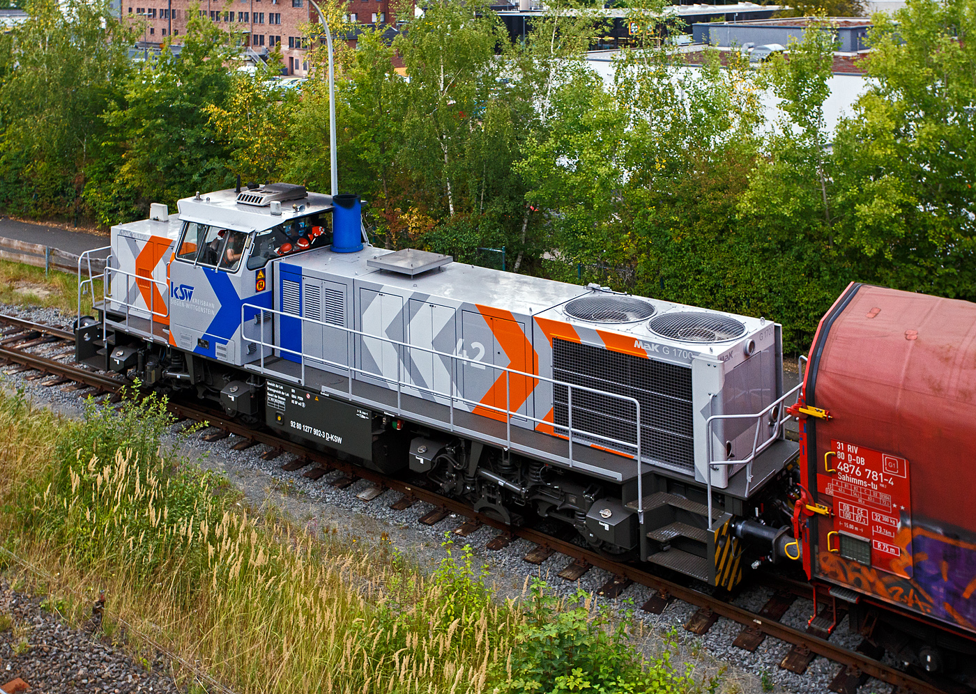 Noch ist die Gleissperre geschlossen, die steht die KSW 42 bzw. 277 902-3 (92 80 1277 902-3 D-KSW) der KSW (Kreisbahn Siegen-Wittgenstein) mit einem Übergabegüterzug auf den KSW Rangierbahnhof Herdorf (Freien Grunder Eisenbahn KSW NE447 / DB-Nr. 9275) zur Abfahrt bereit. Kurzdrauf wird sie mit dem Übergabegüterzug via Betzdorf/Sieg und Siegen nach Kreuztal losfahren.

Die Lok ist eine Vossloh MaK G 1700 BB sie wurde 2001 unter der Fabriknummer 1001108 bei Vossloh gebaut und wurde an die damalige SK - Siegener Kreisbahn GmbH (seit 2004 KSW) geliefert. Sie war die erste gebaute G 1700 BB.

Die Vossloh Lokomotive MaK G 1700 BB ist eine dieselhydraulische Lokomotive. Bei der Überarbeitung der Typenpalette nach der Übernahme der MaK durch Vossloh 1998 wurde im 4. Typenprgramm unter der Bezeichnung G 1700 BB der Bau einer vierachsigen Streckenlokomotive mit Mittelführerhaus vorgesehen. Tatsächlich handelte sich zunächst um eine Weiterentwicklung der G 1206. Die Maschine wurde in einigen Details an die Lokomotiven des 4. Typenprgramms angepasst. Dies betraf in erster Linie den Rahmen, der so geändert wurde, dass die neuen Standarddrehgestelle verwendet werden konnten. Dabei wurden die Rahmenenden so umgestaltet, dass die Lokomotiven die im 4. Typenprgramm üblichen Komfortaufstiege erhielten. Ebenfalls geändert wurde der Dieselmotor. Zum Einsatz kam der schon bei den G 1206 für die Dortmunder Eisenbahn verwendete MTU 12V 4000 R20 mit einer unveränderten Leistung von 1500 kW. Die übrigen Komponenten der G 1700 BB blieben gegenüber der G 1206 weitgehend unverändert.

Die ersten beiden Lokomotiven dieses Typs (die erste für die SK) wurden im Jahr 2001 ausgeliefert. Ende 2002 und Anfang 2003 folgten weitere vier Lokomotiven als Vorratsbauten. Nach diesen sechs Lokomotiven wurde der Bau der G 1700 BB zugunsten der technisch sehr ähnlichen und bereits weiter verbreiteten G 1206 wieder eingestellt.

Bei der G 1700-2 BB handelt es sich nicht um eine Weiteentwicklung der G 1700 BB, sondern um eine komplette Neuentwicklung. 

TECHNISCHE DATEN:
Spurweite: 1.435 mm (Normalspur)
Achsfolge: B´B´  
Länge über Puffer: 14.700 mm 
Drehzapfenabstand: 7.400 mm 
Drehgestellachsstand: 2.400 mm 
größte Breite: 3.080 mm
größte Höhe über Schienenoberkante: 4.220 mm
Raddurchmesser: 1.000 mm (neu)
kleinster befahrbarer Gleisbogen: 60 m
Dienstgewicht: 87,3 t 
Kraftstoffvorrat: 3.500 l
Motor: MTU-12-Zylinder-Dieselmotor, vom Typ 12V4000R20 
Leistung: 1.500 kW bei 1800 U/min
Getriebe: Voith L 5r4 zU2 
Höchstgeschwindigkeit: 100 km/h 
Baujahre: 2001 - 2003 
gebaute Stückzahl: 6 (Ab 2002 folgte die G 1700-2 BB)