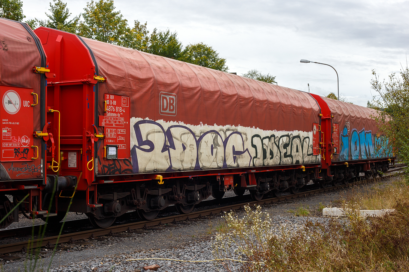 Sechsachsiger Drehgestellflachwagen mit verschiebbarem Planenverdeck und Lademulden für Coiltransporte, 31 80 4876 818-4 D-DB der Gattung Sahimms-tu 900.2, der DB Cargo AG, abgestellt im Zugverband am 26  September 2024 in Herdorf auf dem Rbf der Kreisbahn Siegen-Wittgenstein (KSW).

Der Wagen ist für die Beförderung von schweren witterungsempfindlichen Blechrollen bestimmt. Dieser Spezialwagen besitzt Stirnwände und fünf fest im Untergestell eingebaute Mulden. Die Wagenabdeckung erfolgt mit einer PVC-beschichteten Gewebeplane, die auf 14 Rohrrahmen aufliegt. Zwei auf dem Außenlangträger aufgeständerte Laufschienen dienen als Rollenbahn für mehrere abhebesichere Laufwagen, welche über die Querspriegel mit dem Planendach verbunden sind und dieses somit in Wagenlängsrichtung verschiebbar machen. Die Verriegelung des Planendaches an den Stirnwänden erfolgt über eine Zentralverriegelung, die vom Erdboden aus bedient werden kann. Das Planenverdeck lässt sich an einem Wagenende so weit aufschieben, dass ca. 2/3 der Ladefläche zur zum Be- und Entladen freigegeben werden. Zur Fixierung der Laufwagen des Planenverdecks in der geöffneten Stellung sind an den Endspriegelwagen Festlegeeinrichtungen montiert.

Der Wagen besitzt insgesamt fünf feste Mulden, wobei die beiden äußeren Mulden Coils mit einem Gewicht von maximal 25 t und einem maximalen Durchmesser von 2.250 mm, die mittleren drei Mulden Coils mit maximal 45 t Gewicht bei einem maximalen Durchmesser von 2.700 mm aufnehmen können.

Erstes Baujahr der z.Z. ältesten Wagen war 1984. Hersteller dieser Wagen waren u.a, LHB - Linke-Hofmann-Busch GmbH in Salzgitter, die Waggonfabrik Talbot in Aachen, O&K - Orenstein & Koppel AG in Dortmund-Dorstfeld und die VEB Waggonbau Niesky (DDR). Ursprünglich waren die Wagen mit verschiebbaren Teleskophauben ausgeführt, ab 2018 wurden sie in Wagen mit verschiebbarem Planenverdeck umgebaut.

TECHNISCHE DATEN:
Gattung: Sahimms-tu 900.2
Spurweite: 1.435 mm (Normalspur)
Achsenanzahl: 6 (in 2 Drehgestelle)
Länge über Puffer: 15.000 mm
Drehzapfenabstand: 8.000 mm
Achsabstand im Drehgestell: 3.400 mm (2 x 1.700 mm)
Laufraddurchmesser: 920 mm (neu)
Eigengewicht: 32.300 kg
Höchstgeschwindigkeit: 100 km/h (beladen) / 120 km/h (leer)
Maximales Ladegewichte: 72,5 (ab Streckenklasse D3) / DB 87,5 t (ab CE)
Maximale Tragfähigkeit: 99,5 t
Kleinster bef. Gleisbogenradius: 75 m
Bauart der Bremse: KE-GP (LL)
Bremssohle: IP 116
Handbremse: nein
Intern. Verwendungsfähigkeit: RIV / G1
Hersteller der Plane: Schleswiger Tauwerkfabrik Oellerking GmbH & Co. KG