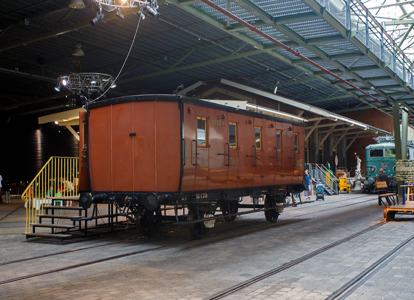 SS C 218
Der zweiachsige dritte Klasse Reisezugwagen SS C 218 „Koekblik“ (Keksdose), Baujahr 1874, der älteste erhaltene Personenwagen der Niederlande, am 24 Juni 2025 im Het Spoorwegmuseum (bis 2005 NSM - Nederlands Spoorwegmuseum / Niederländischen Eisenbahnmuseum) in Utrecht im ehemaligen Bahnhof Maliebaan. Im Museum wird er wegen seiner Form und der Metallverkleidung an der Außenseite „Koekblik“ (Keksdose) genannt. 

Der Wagen wurde 1874 von Beijnes, offiziell Koninklijke Fabriek van Rijtuigen en Spoorwagens J.J. Beijnes (Königliche Fabrik für Kutschen und Eisenbahnwagen J.J. Beijnes) in Haarlem gebaut und an die damalige SS - Maatschappij tot Exploitatie van Staatsspoorwegen (Gesellschaft zum Betrieb von Staatseisenbahnen) geliefert. Er gehörte zu einer Serie von 339 Exemplaren des Typs C5 – 3.Klasse Reisezugwagen mit 5 Abteilen (Serie C 1 - 339) die gebaut wurden. Dieser Wagen ist das einzige noch existierende Exemplar dieser Serie und damit der älteste erhaltene Reisezugwagen der Niederlande. 

Diese Waggons ähnelten noch immer stark an seinen Vorgänger, der Postkutsche. Die Wagen waren nicht besonders komfortabel, der Transport steht im Vordergrund. Er hat keine Heizung, und die Bänke sind aus Holz. Es gibt separate Abteile mit Türen, die nur von außen geöffnet werden können, und einer Außentrittstufe für den Schaffner. Jedes Abteil der 5 Abteile bietet Platz für 10 Personen. Es gibt auch ein separates Damenabteil für allein reisende Frauen. Die Rückenlehnen der Holzbänke reichten nicht alle bis zur Decke. Dadurch konnte der gesamte Waggon mit nur zwei Öllampen beleuchtet werden. 1892 wurde der Waggon mit Gasbeleuchtung ausgestattet, kurz darauf folgte der Einbau einer Heizung, diese wurde mit Dampf aus der Lokomotive betrieben.

Auf dem Dach des Waggons befinden sich wie ein Schweineschwanz geformte Haken sogenannte „Pigtails“, durch die wurde ein Seil, entlang der gesamten Zuglänge geführt, das mit einer Glocke am Tender der Lokomotive verbunden war. So konnte der Schaffner den Lokführer im Notfall warnen bzw. vereinbarte Signale geben. 

Die Wagen waren übrigens nicht alle exakt identisch, es gab Maßabweichungen. Die späteren Wagen hatten mehr Fenster pro Abteil. Jedes Abteil bot Platz für zehn Reisende auf Holzbänken. Es war ein Standardtyp der SS, aber dieselbe Konstruktion fand sich auch bei der NBDS und der NCS. Auch die AB-Wagen (1. und 2. Klasse) verwendeten dasselbe Fahrgestell, aber hier hatten die Reisenden mehr Platz, weil nur vier Abteile auf dem Fahrgestell angebracht waren. Außerdem saßen sie auf gepolsterten Bänken. Die NBDS hatte solche Wagen sogar für die 4. Klasse, die auf der NBDS-Strecke von Gennep nach Gogh in Deutschland im Einsatz waren. Diese einfachen Wagen hatten nur 15 Sitzplätze und 35 Stehplätze. Die 4. Klasse wurde in Deutschland 1928 eingestellt.

Die 1921 noch vorhandenen ehemaligen SS-Wagen erhielten von der NS die Nummern C 2126–2285 (C5). Ab 1917 bilden die Eisenbahngesellschaften HSM beziehungsweise HIJSM (Hollandsche IJzeren Spoorweg-Maatschappij) und SS (Maatschappij tot Exploitatie van Staatsspoorwegen) eine Zweckgemeinschaft, ab 1921 wurde das Rollmaterial, als NS - Nederlandsche Spoorwegen zusammengelegt. Die C 2126 bis 2285 Wagen wurden zwischen 1931 und 1933 größtenteils verschrottet.

Der Wagen SS C 218 wurde jedoch bereits 1922 in einen Dienstwagen des Straßen- und Bauamts mit der Nummer NS 177007 umgebaut. Diese Funktion behielt der Wagen bis 1952. Zunächst drohte ihm auch der Abriss, er wurde jedoch aus historischen Gründen erhalten. Und so blieb er der einzige erhaltene Wagen dieser Serie. Schließlich übernahm die Wagenwerkstatt in Amersfoort den alten Wagen und restaurierte ihn in seinen ursprünglichen Zustand. Nach der Fertigstellung im Jahr 1975 wurde er dem Eisenbahnmuseum gespendet. Im Museum wird sie aufgrund ihrer Form und der Metallverkleidung an der Außenseite „Koekblik“ / „Keksdose“ genannt. 

TECHNISCHE DATEN:
Baujahr: 1874
Hersteller: Beijnes, Haarlem
Gebaute Anzahl: 339 (Typ C5 - 3.Klasse Reisezugwagen mit 5 Abteilen).
Spurweite: 1.435 mm (Normalspur)
Achsen: 2
Länge über Puffer: 9.190 mm
Achsabstand: 4.700 mm
Eigengewicht: 8.960 kg
Höchstgeschwindigkeit: 75 km/h
Abteile: 5 á 10 Personen  
Sitzplätze: 50 (in der 3.Klasse)

Quellen: Het Spoorwegmuseum, nmld.nl und Anschriften
Stand/Überarbeitung: September 2025
