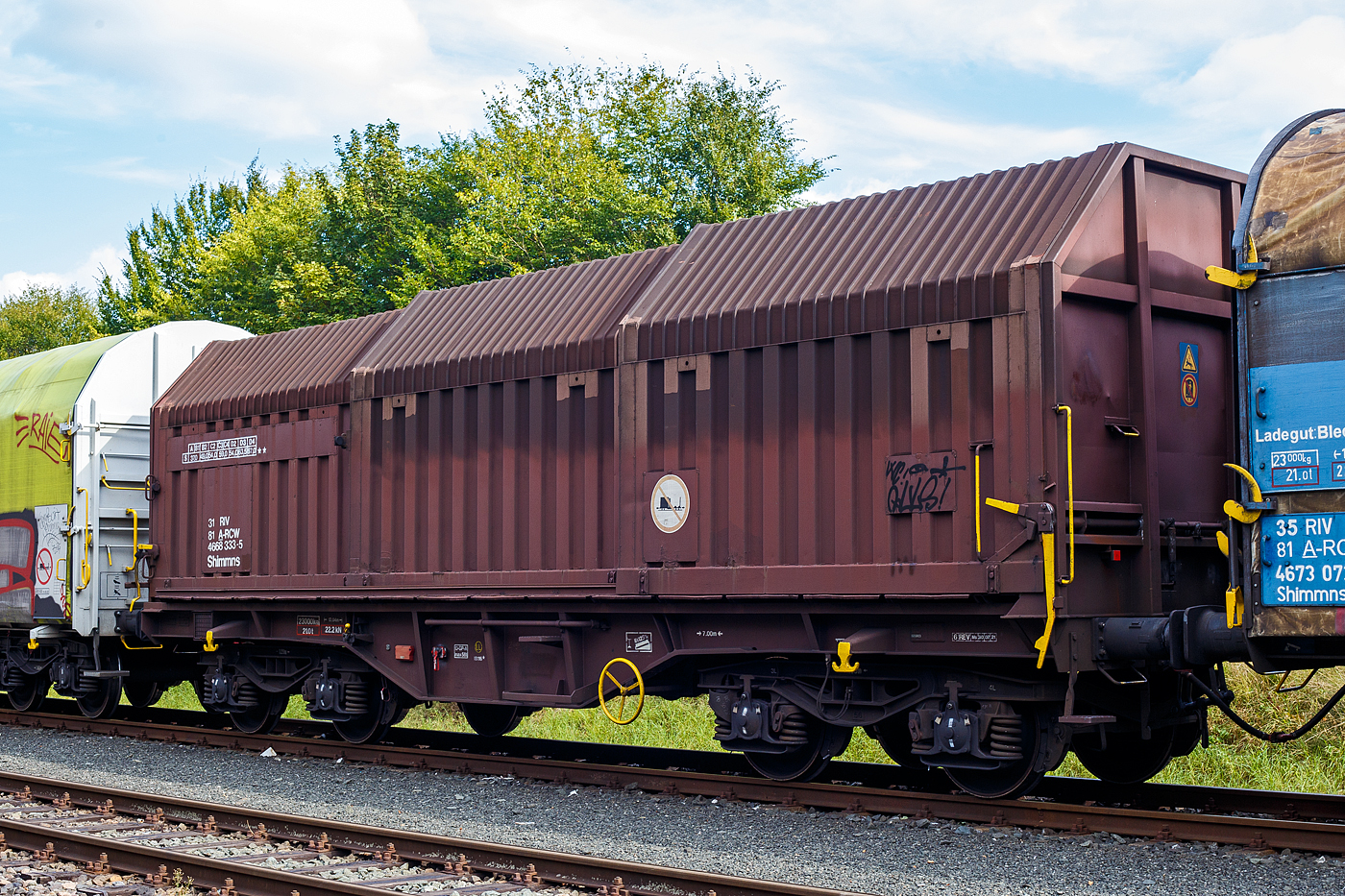 Vierachsiger Drehgestell-Flachwagen mit verschiebbaren Teleskophauben f�r Coiltransporte, 31 81 4665 333-5 A-RCW, der Gattung Shimmns, der Rail Cargo Wagon - Austria GmbH (ein Unternehmen der �BB), abgestellt im Zugverband (�bergabeg�terzug) am 15 September 2024 in Herdorf auf dem Rbf der Kreisbahn Siegen-Wittgenstein (KSW).

Der Wagen wurde 1985 von den Jenbacher Werke AG in Jenbach (�sterreich) unter der Fabriknummer 3.931-334 gebaut.

Der Wagen eignet sich speziell f�r den Transport witterungsempfindlicher Blechrollen. Er besitzt feste Stirnw�nde, 5 fest im Untergestell eingebaute Lademulden und drei teleskopartig angeordnete Hauben, die sich zu beiden Wagenenden hin soweit durch Ineinanderschieben �ffnen lassen, dass jeweils 2/3 des Wagens freigelegt werden kann. Hierdurch ist eine einfache Beladung des Wagens sowohl von oben als auch von der Seite aus m�glich. Der Wagen darf nur mit geschlossenen und verriegelten Hauben verkehren.

Das Untergestell und die festen Stirnw�nde, die als Abkantrahmenkonstruktion mit Blechbekleidung und hutf�rmigen Verst�rkungsprofilen ausgef�hrt sind, sowie die fest auf den Au�enlangtr�gern angeordneten drei Laufschienen je Wagenseite bilden den als Schwei�konstruktion ausgef�hrten Wagenkasten. Das Untergestell mit seinen beiden kr�ftigen Au�enlangtr�gern besteht aus Walz- und Abkantprofilen. Die Untergestellenden bestehen aus den Hauptquertr�gern, den AK-Tr�gern und den Kopfst�cken. Die sattelf�rmig angeordneten, jeweils mit den Au�enlangtr�gern verschwei�ten Quertr�ger bilden zusammen mit 6 mm dicken Muldenblechen die Lademulden.

Die teleskopartig ineinander verschiebbaren Stahlhauben bestehen aus einer Hohlprofil-Rahmenkonstruktion und einer mit Sicken versehenen Beblechung. Die Abdichtung der Hauben untereinander und zu den Stirnw�nden hin erfolgt �ber labyrinthartig ausgef�hrten Umrandungsprofilen. Die Endhauben verriegeln in geschlossener Stellung automatisch. Das Entriegeln der Hauben ist sowohl vom Erdboden wie auch von der Rampe m�glich. Die beidseitig an jeder Stirnwand angeordneten Verriegelungseinrichtungen sind durch Wellen miteinander verbunden. Hierdurch ist das Entriegeln von nur einer Wagenseite aus m�glich. Jede Haube l�uft auf 4 jeweils und mit Abhebesicherungen versehenen Laufwagen mit nadelgelagerten Doppelrollen.

TECHNISCHE DATEN:
Spurweite: 1.435 mm (Normalspur)
Achsanzahl: 4 (in 2 Drehgestelle)
L�nge �ber Puffer: 12.040 mm
L�nge der Ladefl�che: 10.800 mm
Ladebreite (Mulden): 2.400 mm
Drehzapfenabstand: 7.000 mm
Achsabstand im Drehgestell: 1.800 mm
Laufraddurchmesser: 920 mm (neu)
Eigengewicht:  23.000 kg
H�chstgeschwindigkeit: 100  km/h (beladen) / 120 km/h (leer)
Maximales Ladegewicht: 67,0 t (ab Streckenklasse D4) / DB Netz 61,0 t ab CM3
Kleinster bef. Gleisbogenradius: 35 m
Bauart der Bremse: O-GP-A max. 58 t (LL)
Bremssohle: IB 116
Handbremse: Ja
Intern. Verwendungsf�higkeit: RIV