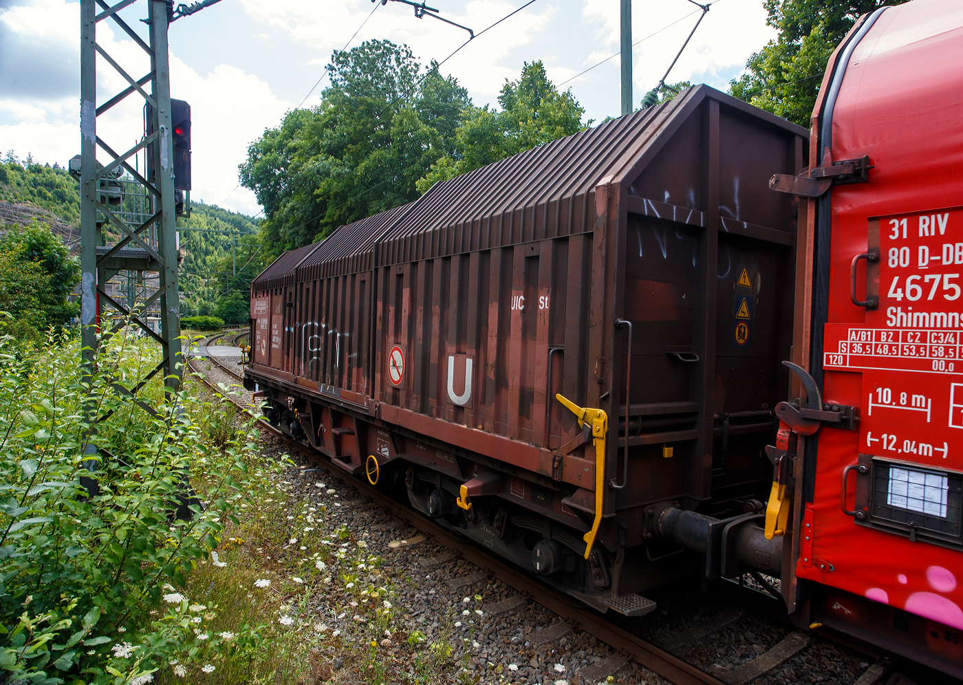 Vierachsiger Drehgestell-Flachwagen mit verschiebbaren Teleskophauben für Coiltransporte, 31 80 4671 011-3 D-DB, der Gattung Shimmns-u 708, der DB Cargo AG, am 16 Juli 2025 im Zugverband (Zug 9921) bei der Zugdurchfahrt in Kirchen (Sieg) in Richtung Siegen bzw. Ziel Kreuztal. 

Der Wagen eignet sich speziell für den Transport witterungsempfindlicher Blechrollen. Er besitzt feste Stirnwände, 5 fest im Untergestell eingebaute Lademulden und drei teleskopartig angeordnete Hauben, die sich zu beiden Wagenenden hin soweit durch Ineinanderschieben öffnen lassen, dass jeweils 2/3 des Wagens freigelegt werden kann. Hierdurch ist eine einfache Beladung des Wagens sowohl von oben als auch von der Seite aus möglich. Der Wagen darf nur mit geschlossenen und verriegelten Hauben verkehren.

Das Untergestell und die festen Stirnwände, die als Abkantrahmenkonstruktion mit Blechbekleidung und hutförmigen Verstärkungsprofilen ausgeführt sind, sowie die fest auf den Außenlangträgern angeordneten drei Laufschienen je Wagenseite bilden den als Schweißkonstruktion ausgeführten Wagenkasten. Das Untergestell mit seinen beiden kräftigen Außenlangträgern besteht aus Walz- und Abkantprofilen. Die Untergestellenden bestehen aus den Hauptquerträgern, den AK-Trägern und den Kopfstücken. Die sattelförmig angeordneten, jeweils mit den Außenlangträgern verschweißten Querträger bilden zusammen mit 6 mm dicken Muldenblechen die Lademulden.

Die teleskopartig ineinander verschiebbaren Stahlhauben bestehen aus einer Hohlprofil-Rahmenkonstruktion und einer mit Sicken versehenen Beblechung. Die Abdichtung der Hauben untereinander und zu den Stirnwänden hin erfolgt über labyrinthartig ausgeführten Umrandungsprofilen. Die Endhauben verriegeln in geschlossener Stellung automatisch. Das Entriegeln der Hauben ist sowohl vom Erdboden wie auch von der Rampe möglich. Die beidseitig an jeder Stirnwand angeordneten Verriegelungseinrichtungen sind durch Wellen miteinander verbunden. Hierdurch ist das Entriegeln von nur einer Wagenseite aus möglich. Jede Haube läuft auf 4 jeweils und mit Abhebesicherungen versehenen Laufwagen mit nadelgelagerten Doppelrollen.

Die Wagen der Bauart Shimmns 708 sind mit 12 verstellbaren Festlegearmen ausgerüstet, die in fest auf den Muldenkappen angeordneten kammartig ausgeführten Halterungen gelagert sind. Die Festlegearme und ihre Anordnung ergeben einen wirksamen Schutz gegen unzulässiges seitliches Verschieben der Coils.

Um unzulässige Radlastabweichungen auszuschließen, ist eine möglichst mittige Verladung der Coils erforderlich. Ein durch alle Mulden in Wagenmitte verlaufender weißer Farbstreifen dient hierzu als Orientierungshilfe.

TECHNISCHE DATEN:
Hersteller: LHB - Linke-Hofmann-Busch GmbH in Salzgitter-Watenstedt
Baujahr: 1972, Fabriknummer 407
Spurweite: 1.435 mm (Normalspur)
Achsanzahl: 4 (in 2 Drehgestelle)
Länge über Puffer: 12.040 mm
Länge der Ladefläche: 10.800 mm
Ladebreite (Mulden): 2.400 mm
Drehzapfenabstand: 7.000 mm
Achsabstand im Drehgestell: 1.800 mm
Laufraddurchmesser: 920 mm (neu)
Eigengewicht: 22.800 kg
Höchstgeschwindigkeit: 100 km/h (beladen) / 120 km/h (leer)
Maximales Ladegewicht: 67,0 t (ab Streckenklasse D4) / 61,0 t bei DB ab CM3
Hier betrug das Ladegewicht laut Laufzettel 65.055 kg
Kleinster bef. Gleisbogenradius: 35 m
Bauart der Bremse: KE-GP (LL)
Bremssohle: IB 116
Handbremse: Ja
Intern. Verwendungsfähigkeit: RIV