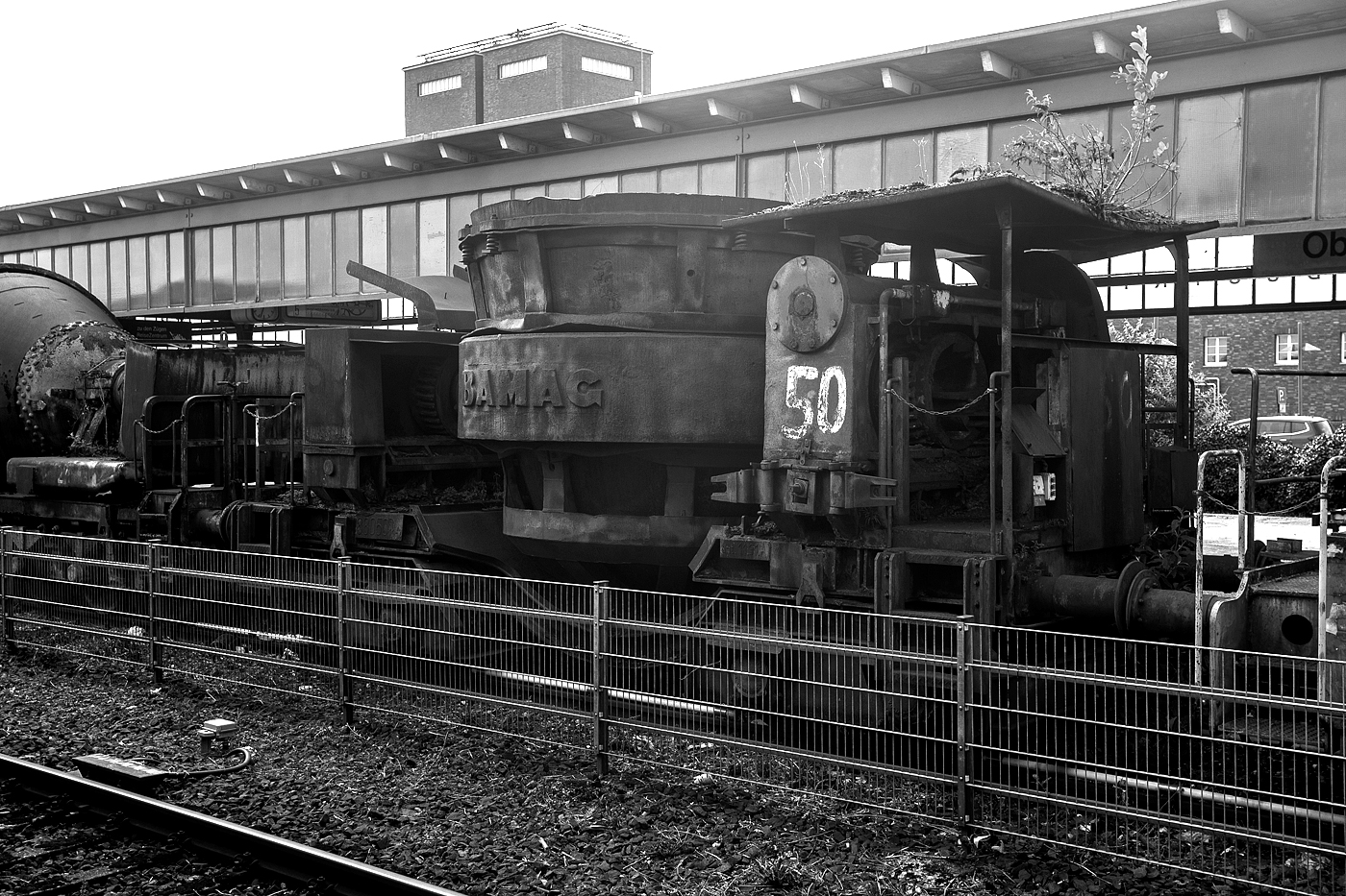 Vierachsiger Schlackenwagen Nr. 50 (Pfannenwagen zum Transport von flüssiger Schlacke) vom Hersteller BAMAG (Berlin-Anhaltische Maschinenbau Aktiengesellschaft) am 24 Juni 2025 beim Hauptbahnhof Oberhausen am Museumsbahnsteig/-gleis. Der Wagen wurde vermutlich auf der Henrichshütte in Hattingen (Ruhr) verwendet. 

Bereits an der massiven und schweren Bauweise sieht man den Fahrzeugen den Verwendungszweck Montanindustrie an. Solche Wagen sind in der Regel nur innerhalb der Stahlwerke im Einsatz, man sieht sie nicht auf den Gleisen der Staatsbahnen.

In Stahlwerken werden solche Wagen für den Transport von Schlacke verwendet. Vor allen bei der Roheisenerzeugung (Eisenverhüttung mittels Hochöfen) fällt sehr viel Schlacke an, aber auch bei der weiteren Stahlerzeugung/-verhüttung. Die flüssige und heiße Schlacke wird u.a. beim Abstich des Hochofens in die Pfanne des Wagens geleitet. Dann wird der Zug mit mehreren Schlackenwagen an die entsprechende Schlackengrube gefahren wo die noch flüssige ausgekippt wird. Nach dem erkalten wird dann die Schlacke mit Radladern o.ä. verlanden und auf die Schlackenhalde verbracht. Dies ist dann der typische Schlackensand oder auch Hüttensand genannt. Hier in der Region dem Siegerland waren viele Hochöfen in Betrieb, heute erinnern meist nur noch die Schlackenhalden daran. Unteranderem auch in Herdorf. 
